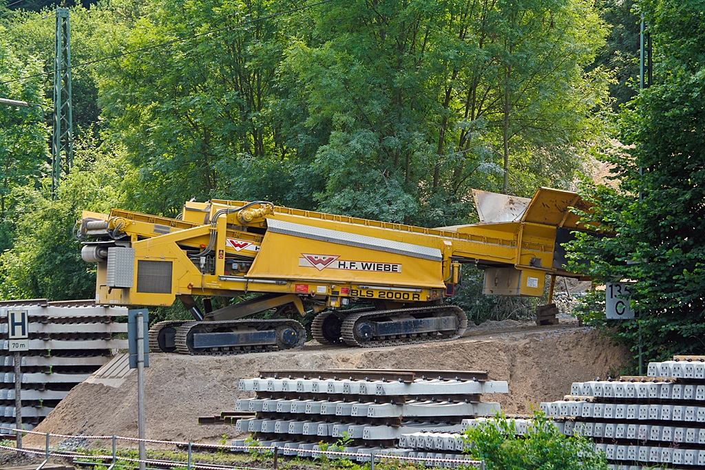 Plasser & Theurer Beladestation BLS 2000 R (mit Raupenfahrwerk) der H.F. Wiebe am 13.07.2013 beim Bahnhof Sinn (Lahn-Dill-Kreis).

Die Beladestation ist zum Verladen von Schotter, Kies und PSS-Material in MSF-Wagen (Materialf�rder- und Silowagen) oder Sch�ttgutwagen, dies ist auch unter vorhandener, abgeschalteter Oberleitung m�glich. Dabei wird Material (z.B. mittels Radlader) in den Schottertrichter aufgegeben. Vom ersten F�rderband auf das zweite hydraulisch ausfahrbares F�rderband transportiert, von dem dann das Material in die entspr. Wagen abf�llt. Der ganze Rahmen ist auf zwei Raupenfahrwerken und ist hydraulisch heb- und senkbar.

Die F�rderb�nder, wie auch die Raupenfahrwerke werden hydraulisch angetrieben, die Energie dazu liefert ein Dieselmotor mit 148 kW Leistung. Die Beladestation wiegt ca. 30.000 kg und ist 12.600 mm lang.
