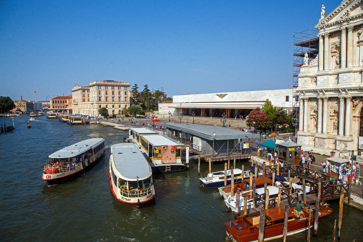 Blick der über den Canal Grande überspannenden Scalzi-Brücke (Ponte degli Scalzi) auf das Empfangsgebäude vom Bahnhof Venezia Santa Lucia am 24.07.2022. 

Der Bahnhof Venezia Santa Lucia der Hauptbahnhof von Venedig. Er ist ein Kopfbahnhof und der einzige Bahnhof der Altstadt von Venedig. Er liegt auf der Insel San Geremia im Sestiere Cannaregio. Seinen Namen hat er von der Santa-Lucia-Kirche, die ursprünglich an diesem Ort stand und für den Bau des Bahnhofs 1861 abgerissen wurde. Ein Gedenkstein auf der Mitte des Platzes erinnert an die ehemalige Kirche.

Neben der Brücke bzw. vor dem Bahnhof liegt die Haltestelle Ferrovia die von Vaporetto (Wasserbuse) und Motoscafi (Wasser-Taxi) bedient werden. An der Haltestelle halten Linien ACTV. Unweit (400 m) in südlicher Richtung (hier im Bild nach links) der Piazzale Roma mit dem Omnibusbahnhof und der Straßenbahnhaltestelle. 

Rechts nur etwas angeschnitten die Scalzi-Kirche (Santa Maria di Nazareth, umgangssprachlich Scalzi), nach der die Brücke ihren Namen bekommen hat.
