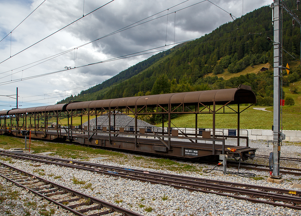 Der MGB Sklv 4811 Autotransportwagen Furkatunnel (Verladewagen) der Matterhorn-Gotthard Bahn, ex FO Sklv 4811 (Furka-Oberalp Bahn), der Serie 4811 bis 4827 für den Transport durch den Furka-Basistunnel abgestellt am 07.09.2021 beim Bahnhof Oberwald (1.365 m ü. M). 

Für die ROLLENDE STRASSE durch den Furka-Basistunnel beschaffte die Furka-Oberalp Bahn 1980 zwei komplette Zugkompositionen jeweils bestehend aus einer Zuglok Ge 4/4 III 81 oder 82, 2 Rampenwagen, 6 Transportwagen (Verladewagen) und 1 Steuerwagen. Seit der Wintersaison 1982 verkehren diese Züge mit hoher Auslastung. Die Gesamtlänge des Zuges beträgt 201 Meter. Um auch Busse und LKW durch den Tunnel transportieren zu können, wurde das entsprechende Wagenprofil der Vollspur von SBB bzw. BLS gewählt. Die maximale, nutzbare Höhe beträgt 4,50 Meter und die Breite 2,70 Meter.

Da die Verladungen auch im Umfahrtunnel Oberwald stattfinden, erhielten die Wagen seitliche Leuchtstoffröhren. Die Wagen sind für die im Tunnel geltende Höchstgeschwindigkeit von 90 Km/h ausgelegt.
Bei Ausfall einer Ge 4/4 III ist es möglich, die Kompositionen auch mit HGe 4/4 I, HGe 4/4 II, Deh 4/4 I oder Deh 4/4 II zu führen. Mitte der Neunzehnhundert-Achtziger Jahre wurde eine komplette Wagengarnitur nachbestellt. Nach Fusion der FO und der BVZ am 1.1.2003 zur Matterhorn-Gotthard Bahn sind alle Fahrzeuge auf die MGB umgezeichnet worden.

TECHNISCHE DATEN:
Vierachsiger Verladewagen (Autotransporter)
FO, Serie Nr. Sklv 4811 - 4822
Anzahl:  12
Baujahr:  1980
Hersteller:  SWS Schieren
Länge über Puffer: 20.900 mm
Gewicht:  41,4 t
Ladegewicht:  20 t
Ladefläche:  53 m²
Maximale Ladehöhe:  3.400 mm
Breite des Wagenkastens:  2.900 mm
Höchstgeschwindigkeit:  90 km/h
Bremsen:  Oe, Laü, mFB, VL

Gezogen werden die Wagen von der eigens für diese Zugkompositionen angeschafften neuen Lokomotiven Ge 4/4 III mit den Nummern 81 und 82, einer Weiterentwicklung der RhB Ge 4/4 II. Eine Zugkomposition sieht folgendermaßen aus:
An der Spitze des Zuges befindet sich immer auf der Seite Realp die Lokomotive. Es folgt ein Rampenwagen (Auffahrwagen) Sklv 4801 bis 4807, dann sechs Verladewagen (Sklv 4811 bis 4827), wieder ein Rampenwagen und als Abschluss ein Steuerwagen (BDt 4361bis 4363) auf der Seite Oberwald. Die Rampen- und Verladewagen sind mit Kuppelstangen fest zu einem Blockzug verbunden. Die Rampenwagen wiederum sind gegen die Lok und den Steuerwagen mit einer automatischen Kupplung +GF+, Typ Brünig, ausgerüstet.

Der Furka-Basistunnel wurde trotz der vielen Querelen im Vorfeld des Baus und der immens hohen Baukosten von 300 Millionen Schweizer Franken ein voller Erfolg. Bis zu Eröffnung des Tunnels fehlte eine Winterverbindung. Jedes Jahr musste die Steffenbachbrücke auf der FO-Strecke im Herbst abgebaut und im Frühjahr wieder aufgebaut werden. Das Goms war in diesen 8 Monaten, solange war der Verkehr der FO vor dem Tunnelbau unterbrochen, mehr oder weniger vom Leben abgeschnitten. Es drohte die Entvölkerung, da auch geeignete Arbeitsplätze fehlten. In den Wintermonaten versah die Furka-Oberalp Bahn hauptsächlich Pflichtaufgaben, die fast nur noch sozialen Charakter hatten. Seitdem der Tunnel in Betrieb ist, hat sich die Situation grundlegend geändert. Das Goms und das ganze Wallis sind besser erreichbar, und der Wintertourismus hat Einzug gehalten. Generell wirkte sich die Eröffnung des Furka-Basistunnel positiv auf alle von der Furka-Oberalp Bahn bedienten Gebiete aus. Und die Autransport-Züge sind nicht mehr wegzudenken. Bis zu 180.000 Autos jährlich befördern die beiden Autotransportzüge durch den Furka-Basiatunnel.

Technische Daten Furka-Basistunnel:
Länge Oberwald bis Realp:  15,407 km
Maximale Steigung: 17,5 Promille
Höhendifferenz Portal Oberwald - Portal Realp:  160 m
Kreuzungsstellen im Tunnel:  2
Nutzlängen der Kreuzungsstelle:  je 500 m
Bauzeit:  1973 - 1982
Vorgegebene Durchfahrtsgeschwindigkeit: 55 - 75 km/h
Maximale Durchfahrtsgeschwindigkeit:  90 km/h
Fahrzeit:  15 - 22 Minuten

Der Furkatunnel ist 15,4 Kilometer lang und verbindet die Stationen Realp und Oberwald. Die einspurige Tunnelstrecke wird durch zwei je 774 Meter lange Tunnel-Ausweichen unterteilt. Der automatische Block erlaubt eine Zugfolge von 8 bis 10 Minuten.
