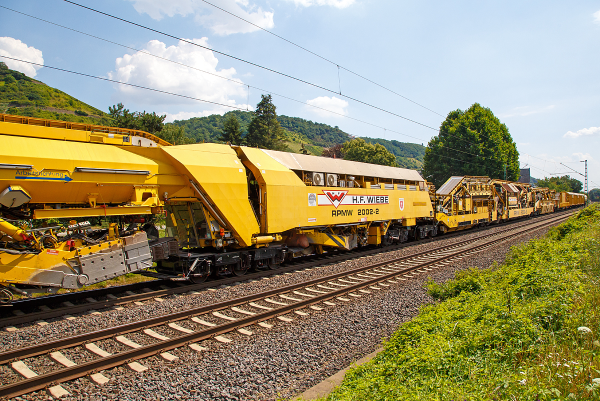 
Die Wiebe Recycling-Planumsverbesserungsmaschine mit Schotterrecycling RPMW 2002-2 am 14.07.2018 im Zugverband auf der Fahrt durch Leutesdorf,  auf der KBS 465 (Rechte Rheinstrecke) in Richtung Norden. 

Über 140 Meter lang, knapp 5 Meter hoch und gute 560 Tonnen schwer – die Gleisbaumaschine RPMW 2002-2 gehört zu den größten der Welt. Mit ihrer beachtlichen Leistung und dem umfänglichen Leistungsspektrum ist sie ein äußerst wichtiger Bestandteil des Maschinenparks der Unternehmensgruppe Wiebe.

Die Recycling Planumsverbesserungsmaschine führt verschiedene Gleisbauarbeiten durch – und das mit einer Motorengesamtleistung von über 2100 Kilowatt. Diese Kraft kommt bei Totalsanierungen zum Tragen, bei denen die RPMW 2002-2 mehrere Arbeitsschritte simultan ausführt: Die verschmutzte Schotterbettung wird ausgebaut, das abgetragene Bettungsmaterial aufbereitet, Schotter recycelt und die Unterbauplanumsschicht erneuert.

TECHNISCHE DATEN:
Hersteller: Plasser & Theurer
Baujahr, Erstzulassung: 2000
Spurweite: 1.435 mm
Anzahl der Achsen: 29, davon 12 angetrieben
Baumaschinennummer: Aushubmaschine 3369, Recyclingmaschine 3368, Antriebswagen 3367,  Stopf- u. Einschotterungsmaschine 3370
Motoren: Caterpillar 
Leistung: 2.860 PS
Gesamtlänge über Puffer: 140,90 m
Breite: 3,29 m
Höhe: 4,65 m
Gewicht: 560 t
V zul. im Zug eingestellt: 100 km/h
V max. bei Eigenfahrt: 20 km/h
Fahrradius: 120 m

Der große Unterschied zwischen Planumsverbesserungsmaschine und Bettungsreinigungsmaschinen besteht darin, dass die Planumsverbesserungsmaschine nicht nur den Schotter ausheben und recyceln kann, sondern auch einen Teil des Planums (Unterbau) ausheben (bis 1.200 mm unter SOK). Zudem kann die RPM vor der Neuschotterung (Recycling- mit Neumaterial) auch eine Planumsschutzschicht (PSS-Material) einbringen.
Der Neuschottern wird durch die Stopfeinrichtung 09-32 Stopfsatellit im Gleisbett verdichtet und das Gleis ist danach wieder mit 70 km/h befahrbar.

Hier in der Bildmitte die Schotteraushubkette:
Die Schotteraushubkette startet den Arbeitsprozess mit der Aufnahme der oberen Schotterschicht, dem recycelbaren Anteil der alten Bettung. Die Räumtiefe lässt sich exakt einstellen. So passt sich die Planumsverbesserungsmaschine den Anforderungen verschiedener Gleisanlagen an und gewährleistet eine effiziente Gewinnung wiederverwendbaren Schotters.
Dank der direkten Materialbewegung innerhalb der Maschine gelangt das abgetragene Material ohne Umwege zum Sternsieb, wo eine Trennung des bindigen Materials vom Schotter stattfindet, dort kommt auch eine  Hochdruckwassersprühanlage zum Einsatz, die eine noch bessere Reinigung des Schotters gewährleistet. Anschließend werden Metallteile über den Metallabscheider entfernt, woraufhin der gereinigte Schotter zur Brechanlage gelangt, mit ihr wird das Material angeschärft. Die so geschaffenen scharfen Kanten garantieren ein Verkeilen der einzelnen Schottersteine zu einer stabilen Gleisunterlage. Eine freischwingende Vibrationssiebanlage siebt die geschärften Schottersteine erneut und trennt sie von Kleinanteilen. Diese werden zusammen mit dem Abraum zur Maschinenfront gefördert. 
Gesäubert und geschärft gelangt das Material über die Förderanlagen zu den Einschottervorrichtungen auf der Stopf- und Einschotterungsmaschine, die den recycelten Schotter als unterste Lage einbringt und verteilt. Der Schoppbalken verdichtet die neu aufgetragene Schotterschicht, das integrierte Stopfaggregat stabilisiert sie. Für die wichtigen Ausfädelstellen und zur Vermeidung von Engpässen wird in doppelstöckigen MFS Neuschotter mitgeführt.