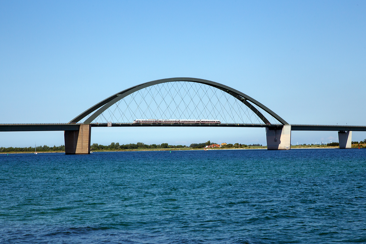 
Ein ICE TD (Baureihe 605) bzw. VT 605, fährt am 15.06.2015 über die Fehmarnsundbrücke in Richtung Festland, aufgenommen von  Grossenbroder Fähre. 

Die Fehmarnsundbrücke ist eine kombinierte Straßen- und Eisenbahnbrücke, die die Insel Fehmarn in der Ostsee mit dem Festland bei Großenbrode verbindet, sie wurde 1963 eröffnet.

Die 963 Meter lange Brücke überspannt den 1300 Meter breiten Fehmarnsund, wobei die restlichen 337 Meter aus beidseitigen Rampen bestehen. Sie hat eine lichte Höhe von 23 Metern über dem Mittelwasser und bietet für den Schiffsverkehr einen Durchgang von 240 Metern Breite sowie eine Durchfahrtshöhe von 23 Metern über NN. Sie ist eine Stahlkonstruktion mit 21 Metern Breite, von denen sechs Meter von der Deutschen Bahn genutzt werden. Der zirka 268,5 Meter lange Bogen hat eine Stützweite von 248 Metern und ist mit 45 Metern über der Fahrbahn höchster Punkt.

Nach einer Festen Fehmarnbeltquerung (welche geplant ist) wird die vorhandene Fehmarnsundbrücke den höheren Belastungen nicht mehr genügen könnte.