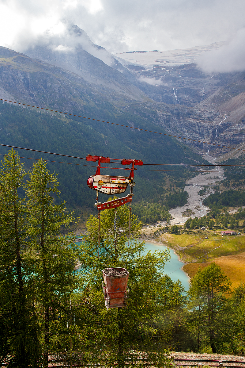 Unterhalb Alp Grüm werden die Alp Grüm der Berninabahn erneuert, daher gibt es eine Materialseilbahn von Alp Grüm hinab zu den Galerien, hier am 06.09.2021. So wird oben in Alp Grüm der Beton gemischt und dann mit der Seilbahn hinab gefahren. 

Die hier verwendete Bahn ist vom italienischen Seilbringungs-Spezialisten Seik (Truden). Der verwendete Wagen ist ein Seilkran vom Typ SFM 30/60.  In dem Seilkran befindet sich ein Verbrennungsmotor der das Hubwerk antreibt (dieser läuft nur bei der Hubbewegen) um wie hier den Betonkübel anzuheben und später wieder abzusenken. Die Hubkraft beträgt im Einzelzug 3t oder wie hier im Doppelzug mit dem Waagbalken 6t. Das Gesamtgewicht beträgt ca. 1t. Die Winde für das Zugseil kann nach Belieben (Bergseite – Talseite) platziert werden.