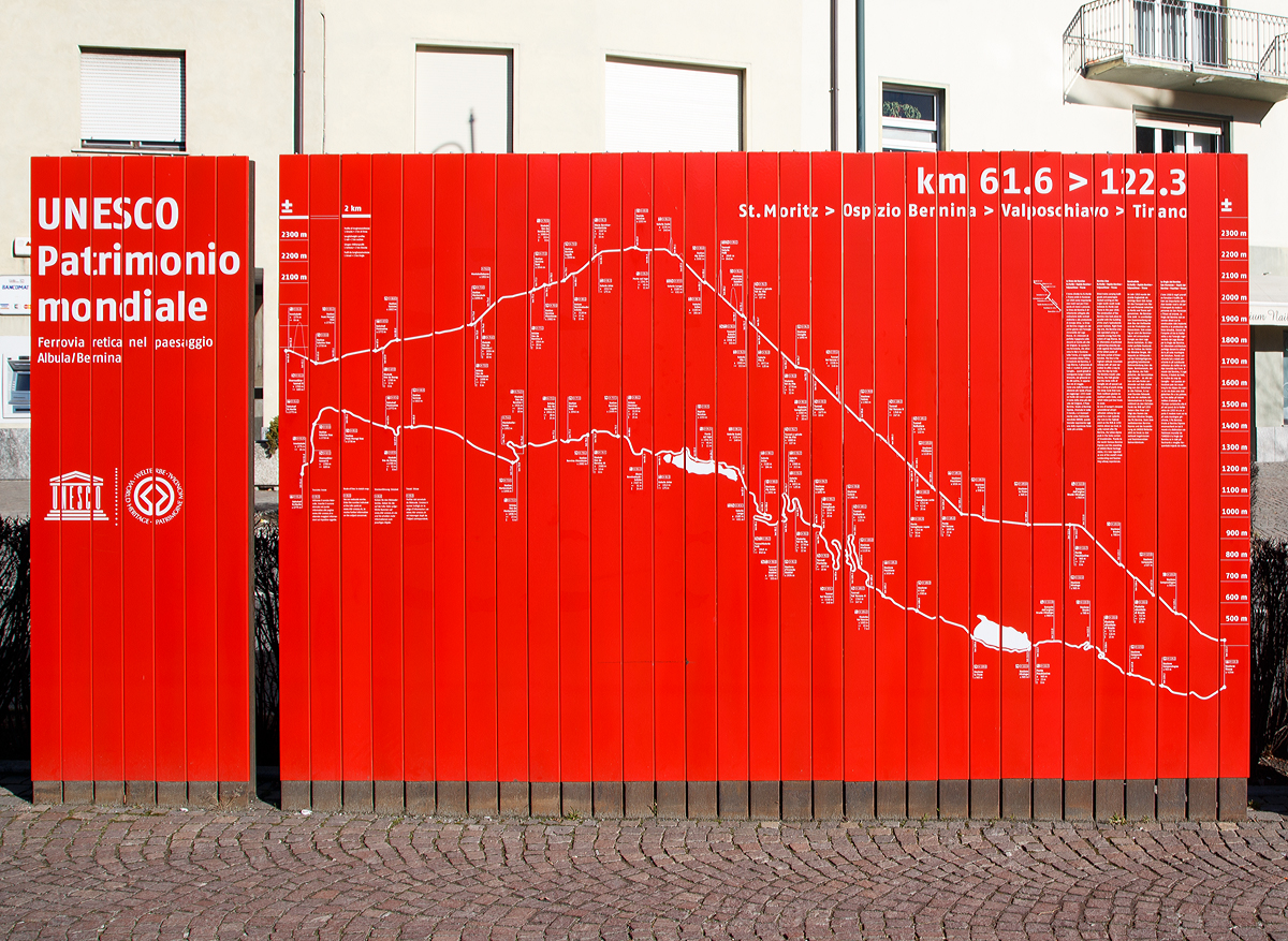 
Werbeschild für das UNESCO-Weltkulturerbe Berninabahn in Tirrano (19.02.2017).
Oben sieht man sehr gut das Steigungsdiagramm und darunter den Streckenverlauf. Eine Stehle entspricht 2 km.

Die Berninabahn, auch Berninalinie genannt, ist eine eingleisige meterspurige Eisenbahnstrecke der Rhätischen Bahn (RhB) und war bis zum Zweiten Weltkrieg eine eigenständige Bahngesellschaft (abgekürzt BB). Die Gebirgsbahn verbindet den Kurort St. Moritz im Schweizer Kanton Graubünden über den Berninapass (Ospizio Bernina  2.253 m ü. M.) mit der italienischen Stadt Tirano. Sie gilt als höchste Adhäsionsbahn der Alpen und – mit bis zu sieben Prozent Neigung – als eine der steilsten Adhäsionsbahnen der Welt.

Die Berninabahn wurde gemeinsam mit der Albulabahn am 7. Juli 2008 in die Liste des UNESCO-Weltkulturerbes aufgenommen.  

Daten der Strecke:
Streckenlänge : 60,688 km
Spurweite: 1.000 mm (Meterspur)
Stromsystem: 	1000 Volt DC
Maximale Neigung: 	70 ‰
Minimaler Radius: 	45 m

In St. Moritz beginnt die Berninabahn. Die Bahn verlässt den Bahnhof in Richtung Osten und überquert auf einem 64 m langen Viadukt den Inn. Danach führt sie durch den 689 m langen Charnadüra-Tunnel II, den längsten Tunnel der gesamten Strecke. Die folgende Station Celerina Staz ist mit 1716 m ü. M. der niedrigste Punkt auf der Nordseite des Berninapasses. Bis Ospizio Bernina wird die Linie nun fast ununterbrochen ansteigen. 

Der Bahnhof in Pontresina weist zusammen mit dem Bahnhof St. Moritz die Besonderheit auf, dass zwei verschiedene Stromsysteme innerhalb des RhB-Netzes zusammentreffen. Hier beginnt für mich eigentlich der schönste Abschnitt der Berninabahn über Ospizio Bernina auf 2.253 m ü. M. bis Alp Grüm (2.091 m ü. M.). Wobei hinter dem Bahnhof Alp Grüm dann der steilste Abschnitt der Strecke beginnt. Von hier aus klettert die Linie mit einem Gefälle von bis zu 70 ‰ über mehrere Kehren ins Puschlav hinunter. Da dies ohne Zuhilfenahme einer Zahnstange geschieht, gehört die Berninabahn mit zu den steilsten Adhäsionsbahnen.

