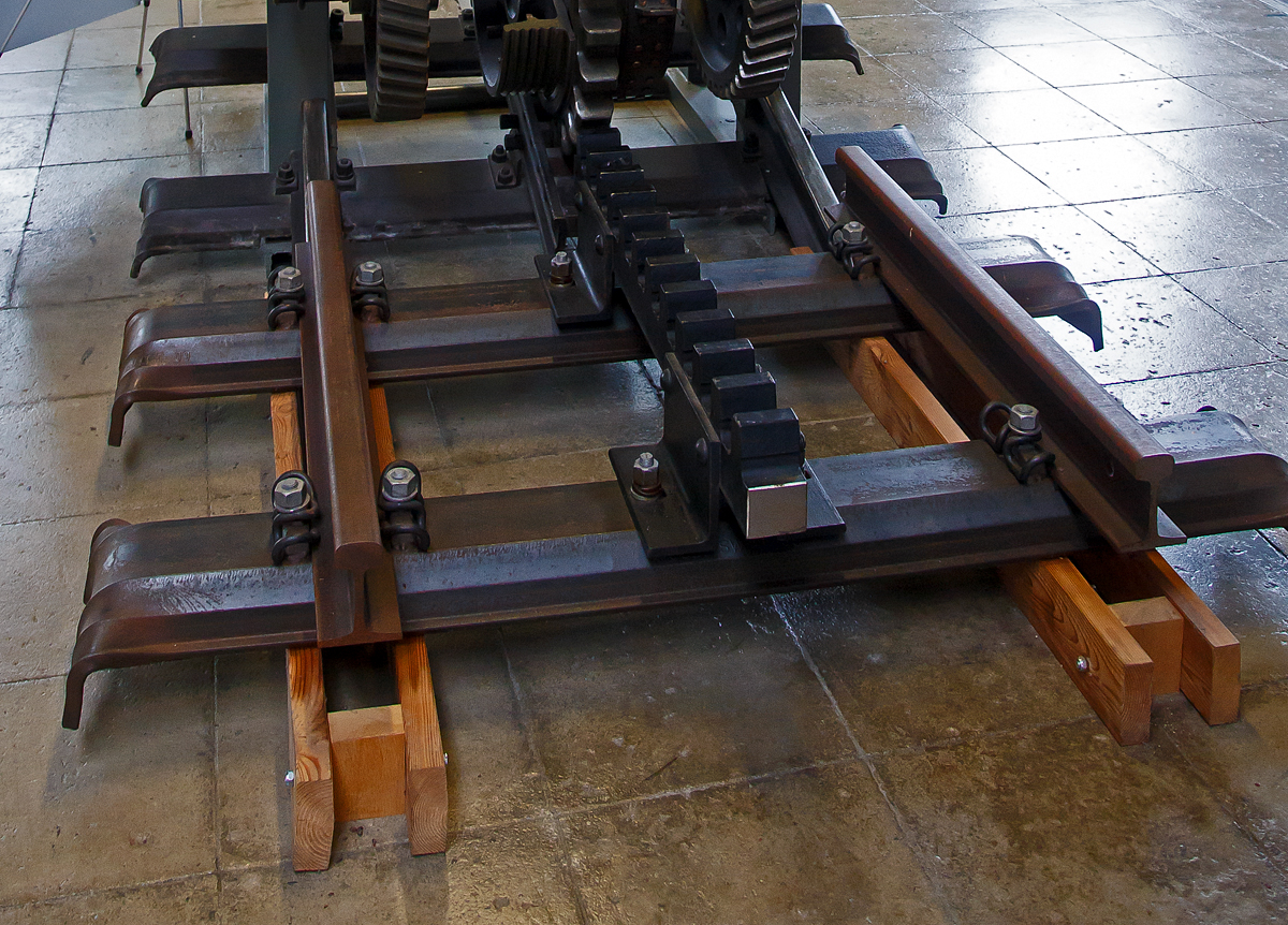 Zahnradbahn Schienenst�ck als Meterspur-Gleis mit dem System von Roll (heute Tensol), montiert auf Stahlschwellen (Trogschwellen),  am 11.09.2022 in der Lokwelt Freilassing. Dahinter kann man ein wenig eine nach dem System Riggenbach der Zugspitzbahn erkennen.

Die von der Schweizer Firma Von Roll (heute Tensol) entwickelte Zahnstange ist nur dem Namen nach eine Lamellenzahnstange, n�mlich eine einlamellige. Beim System von Roll, war eigentlich kein gro�er Erfinder am Werk. Die Idee bei diesem System war, die vorhandenen Zahnstangen von Emil Strub und Niklaus Riggenbach zu erg�nzen. Letztlich war das System so gut, dass es sich als eigenes System durchsetzen konnte und sehr oft als Erg�nzung dient.

Die Verzahnungsgeometrie (Zahnteilung 100 mm) entspricht denen der Systeme von Strub und Riggenbach und ist so kompatibel, darum kann sie bei Bedarf verwendet werden. Sie hat dieselbe Zahnteilung von 100 mm (Verzahnungsgeometrie) wie die Zahnstangen nach Riggenbach und nach Strub. Von letzterer unterscheidet sie sich nur in der Grundform, einfaches Breitflach-Profil anstatt einer Keilkopfschiene. So kommt diese Zahnstange in erster Linie bei Neubauten sowie als preisg�nstiger Ersatz alter Zahnstangen nach den Systemen Riggenbach oder Strub zur Anwendung. Sie ist zwar dicker (30–80 mm, je nach Zahndruck) als eine der Lamellen nach Abt, doch ihre Biegsamkeit ist ausreichend, um flexibler als die Originale nach Riggenbach oder Strub anwendbar zu sein. Sie kann auch durchgehend verschwei�t werden. Zur Befestigung auf den Schwellen dienen besondere Profilstahl-S�ttel.

Man vereinfachte bei von Roll die Zahnstangen von Niklaus Riggenbach und Emil Strub. Statt einer Leiterzahnstange oder einer Keilschiene, verwendete man bei von Roll ein einfaches flaches Eisen. Diese L�sung reduziert die Kosten bei der Fertigung der einzelnen Elemente und lie� eine einfachere Bearbeitung der Zahnstange zu. Zudem wollte man die Probleme mit den Weichen l�sen. Da die Weichen f�r Zahnrad ausgesprochen kompliziert waren, erachtete man bei „von Roll“ eine Vereinfachung als gro�en Vorteil. Im Bereich von Weichen war die Zahnstange von Roll mit ihren geraden flachen Eisen viel einfacher zu montieren. Damit wollte man die komplizierten Weichen nach Riggenbach ersetzen. Es war daher nie geplant auch Strecken damit auszur�sten. 
