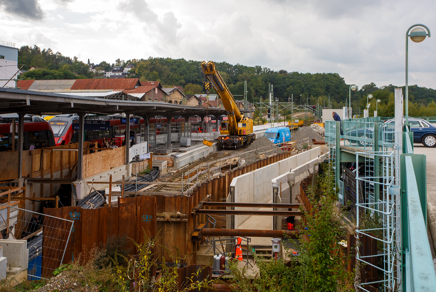 10 Jahre ist es nun her - Daran krankt es in Deutschland. Nun ist es 10 Jahre her wo eigentlich wieder das Gleis 107 im Bahnhof Betzdorf/Sieg aktiv sein sollte. Eigentlich war das Gleis 107 im Jahr 2017 auch fertig und 2019 hatte es dann auch eine Oberleitung, wobei für die Dieseltriebwagen der HLB die von hier fahren sollten man sie nicht gebraucht hätte. Dann war den Verantwortlichen doch aufgefallen das man was vergessen hat, denn wie kommt man nun barrierefrei vom P+R Parkhaus (Park & Ride) zu den Bahnsteigen😉. Den Weg hatte man nun mit dem wieder aktiven Gleis verbaut. 

So kam dann auf die Idee die Personen-Unterführung (Personentunnel) unter Gleisen auch unter Gleis 107 und zum Parkhaus zu verlängern. Dafür wurde auch wieder ein Teil vom Gleis demontiert, Spundwand mittels eingerammter Spundbohlen erstellt und eine Baugrube ausgeschachtet. Die barrierefreie Rampe ist nun schon länger fertig betoniert. 

Aber hier müssen wohl noch unter dem P+R Parkhaus bis zur Sieg (Fluss) Horizontalbohrungen gemacht werden, für erforderliche Regenwasser-Entwässerungs-Leitungen. Dazu ist auch hier der 125 t KIROW Gleisbauschienenkran KIROW Multi Tasker KRC 810 T, D-HGUI 99 80 9419 024-1, der HERING Bahnbau GmbH (Burbach) hier im Einsatz. Er hebt die Bohrrohre in die Baugrube und Kübel mit dem Abraum aus der Baugrube aus der Baugrube. Eigentlich ist ein 125 t Kran damit unterfordert (überdimensioniert), aber man muss bedenken es wurde eine weite Auskragung (Ausladung vor Puffer) und das freistehend (Nichtabgestützt) benötigt. 

Ob das ganz bis zum Fahrplanwechsel im Dezember 2025 fertig wird, wage ich zu bezweifeln, denn nun schreiben wir schon den 30. September 2025.
