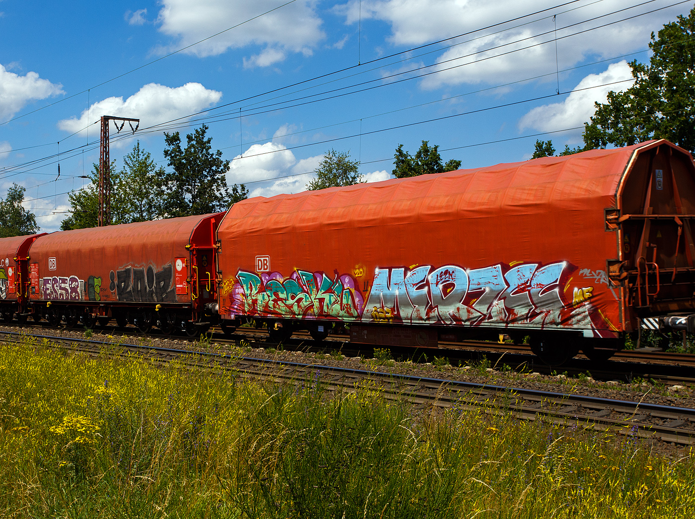 2-achsiger Flachwagen mit öffnungsfähigem Planendach und Stoßverzehreinrichtung zum Schutz vor Rangierstößen, 41 80 3384 088-5 D-DB, der Gattung Kijls 450, der DB Cargo AG (leider wie viele solcher Wagen sehr beschmiert), am 11 Juni 2024 im Zugverband bei einer Zugdurchfahrt durch Rudersdorf (Kreis Siegen) in Richtung Siegen bzw. Kreuztal.

Der Wagen wurde 2001 von ALSTOM LHB (vormals Linke-Hofmann-Busch GmbH in Salzgitter-Watenstedt gebaut.

Der Wagen lässt an sich einiges abprallen, er ist mit einer Stoßverzehreinrichtung zum Schutz vor Rangierstößen ausgestattet. So eignet sich dieser Wagen besonders zum Transport von stoßempfindlichen Gütern (z. B. Komponenten für die Automobilindustrie, Motoren, Getriebe, Maschinen, Trafos). Aufgrund des geschlossenen und verriegelten Planendachs gewährt er auch Schutz für nässeempfindliche Güter. Zusätzliche Zug-Spannsysteme gewährleisten eine schnellere Ladesicherung. Bei einer Ladelänge von über 14 Meter bietet der Wagen einen Laderaum von 109 m³.

Der Wagen besitzt einen unter dem Wagenkasten durchlaufenden, hydraulischen Stoßbalken, der die im Wagen befindlichen Güter weitgehend vor Rangierstößen schützt. Dadurch wird die auf die Ladung wirkende rechnerische Beschleunigung bei Auflaufgeschwindigkeiten bis 9 km/h auf 1 g (9,81 m/s²) begrenzt.

Die Stirnwände bestehen aus zwei schrägen Stirnwandsäulen, zwei Querträgern und einem Rahmen, der gleichzeitig das Dichtsystem aufnimmt. Die Dicke der Beblechung beträgt 2 mm. An den Stirnwänden sind die robusten 4-Punkt-Zentralverriegelungen angeordnet, mit der das Planendach zuverlässig gesichert werden kann. Die Betätigungseinrichtungen sind so angeordnet, dass das Ent- und Verriegeln des Planendaches sowohl vom Erdboden als auch von einer Rampe aus möglich ist. Die insgesamt 4 Verriegelungshaken (je Längseite) einschließlich der beiden Betätigungshebel sind über drehbar gelagerte Wellen untereinander verbunden. Dadurch ist ein Betätigen der Zentralverriegelung von jeder Wagenseite aus möglich.

Das Planendach besteht aus 2 End- und 10 Mittelspriegeln mit einer PVC-Beschichteten Gewebeplane. Die Spriegel ruhen auf Laufwagen, die wiederum selbst auf an den Langträgern angeordneten Fahrschienen stehen. Unter die Fahrschienen greifende Rollen an den Laufwagen wirken als Sicherungen und verhindern so ein Abheben der Hauben bei Auflaufstößen oder Windbelastung.

Das Planendach lässt sich mit geringem Kraftaufwand jeweils zu einem Wagenende hin verfahren und so weit zusammenschieben, dass ca. 3/4 der Ladelänge zur uneingeschränkten Beladung zur Verfügung stehen.

TECHNISCHE DATEN des Wagens:
Gattung: Gattung Kijls 450 (Gattungskennzahl 3384)
Erstes Baujahr: 2001
Spurweite: 1.435 mm (Normalspur)
Achsanzahl: 2
Länge über Puffer: 16.500 mm
Achsabstand: 10.000 mm
Ladelänge: 14.150 mm
Ladebreite: 2.850 mm
Ladefläche: 40,3 m²
Laderaum: 109 m³
Fußbodenhöhe über SOK: 1.2430 mm
Höchstgeschwindigkeit: 100 km/h (beladen) / 120 km/h (leer)
Eigengewicht: 17.450 kg
max. Zuladung: 27,5 t (ab Streckenklasse D) / DB Netz CM 24,5 t
Bauart der Bremse: KE-GP-A
Handbremse: nein
Intern. Verwendungsfähigkeit: nein
