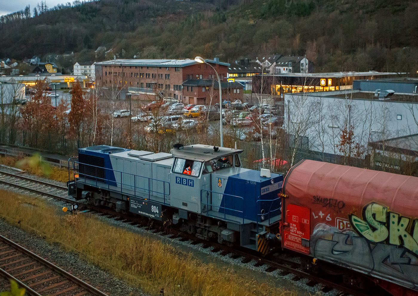 Aktuell besteht bei der KSW (Kreisbahn Siegen-Wittgenstein) immer noch Lokmangel, so ist zurzeit auch die RBH 804 / 275 804-9 (98 80 0275 804-9 D-RBH) eine MaK G 1206 der RBH - RAG Bahn und Hafen GmbH (Gladbeck), an die KSW vermietet. Hier fährt sie am 08 Dezember 2025 um 16:21 Uhr, beim allerletzten Licht des Tages, mit einem Übergabegüterzug (leere Wagen) auf dem Rbf der KSW in Herdorf (Betriebsstätte Freien Grunder Eisenbahn - NE 447) vor zur Gleisspeere (Anschlussstelle) zur Hellertalbahn. Die Helligkeit des Bildes täuscht etwas über die eigentlichen tatsächlichen Lichtverhältnisse hinweg.

Die MaK G 1206 wurde 1997 von der Siemens Schienenfahrzeugtechnik (SFT) in Kiel-Friedrichsort unter der Fabriknummer 1000903 gebaut und an die damalige RAG - Ruhrkohle AG, RAG Bahn- und Hafenbetriebe in Gladbeck als RAG 804 ausgeliefert, 2004 erfolgte die Umfirmierung in RAG Bahn und Hafen GmbH (RBH) und nach der Übernahme durch die DB Schenker Rail Deutschland AG (heute DB Cargo) erfolgte 2006 die Umfirmierung in RBH Logistics GmbH. 

Die SFT war die Nachfolgefirma der Krupp Verkehrstechnik GmbH bzw. der früheren Maschinenbau Kiel GmbH (MaK). Die SFT wurde am 1. Oktober 1998 von Siemens an die Vossloh AG verkauft, in Vossloh Schienenfahrzeugtechnik (VSFT) umbenannt und firmierte seit 23. April 2004 als Vossloh Locomotives GmbH. Die Vossloh-Lokomotiven aus Kiel trugen das Kürzel MaK in der Fahrzeug-Typenbezeichnung. Nach der Fokussierung von Vossloh auf den Bereich Schiene erfolgte 2019 ein weiterer Verkauf an die Chinesische CRRC Zhuzhou Locomotive Co., Ltd.

Die Lok (MaK G 1206):
Die MaK G 1206 ist im schweren Rangier- und Streckendienst einsetzbar. Sie wurde 1997 zum ersten Mal gebaut, erster Kunde war die RAG, somit diese (die 804) erst die Vierte der Serie. Bis 2016 wurden insgesamt 323 Fahrzeuge der Baureihe G 1206, in 5 verschiedenen Varianten, produziert. Sie wurden nach Deutschland, Österreich, der Schweiz, Luxemburg, Italien, Spanien, den Niederlanden und nach Frankreich verkauft worden. Allein 23 Lokomotiven fahren als BB 61000 bei der SNCF, davon sechs mit MTU-Motor (Die G 1206 gibt es wahlweise mit Caterpillar- oder MTU-Motor). 

Diese Lok der ersten Serie (von 11 Stück) hat einen wassergekühlten 90°V16- Zylinder-Viertakt-Dieselmotor mit Abgasturbolader und Ladeluftkühlung, sowie mit Direkteinspritzung, vom Typ MTU 16V 396 TC 14 des Herstellers MTU Friedrichshafen GmbH (MTU steht für Motoren- und Turbinen-Union). Der Motor hat ein Zylinderbohrung von 165 mm und einen Kolbenhub von185 mm, damit hat der Motor einen Hubraum von 63.3 Litern. Die Motorleistung beträgt 1.570 kW (2.135 PS) bei 1.800 U/min, solche Leitungen erbringe heute 12 Zylindermotoren. Das Strömungsgetriebe ist ein Voith L 5r4 zU2. Die Höchstgeschwindigkeit der Lok beträgt 80 km/h.

Die G 1206 nimmt im 3. Typenprogramm eine besondere Rolle ein. Sie ist nicht nur die größte und leistungsstärkste Mittelführerhaus-Lok als Schlusspunkt dieser Typenreihe, sondern hat mit ihrer Verbreitung in vielen Ländern Europas inzwischen eine gebaute Stückzahl erreicht wie noch keine unabhängig von Staatsbahnaufträgen entwickelte Standardlok aus Kiel vor ihr. Was 1997 als neue Lokgeneration für die Ruhrkohle AG begann, ist heute eine Lokomotive, die in acht europäischen Ländern bei zahlreichen Bahnen, darunter zwei Staatsbahnen, im Einsatz steht.

Mitte der 1990er Jahre entstand bei den Bahn- und Hafenbetrieben der Ruhrkohle AG Bedarf nach neuen Diesellokomotiven. Diese Maschinen sollten nicht nur im schweren internen Rangier- und Zustelldienst eingesetzt werden, sondern auch - die Möglichkeiten der Liberalisierung nutzend - mit schweren Kohlezügen auf Strecken der DB AG verkehren. Dafür benötigten die Lokomotiven eine entsprechend hohe Motorleistung und eine Höchstgeschwindigkeit, die einen freizügigen Einsatz auf DB-Gleisen zulässt.

Den Auftrag zur Entwicklung und Lieferung von elf dieser Lokomotiven bekam die damalige Siemens Schienenfahrzeugtechnik GmbH. Das Ergebnis war die G 1206, deren erstes Exemplar Ende Juli 1997 an die RAG ausgeliefert wurde. Sie ist aus der G 1205 entwickelt worden und unterscheidet sich von dieser hauptsächlich durch den Dieselmotor mit 16 statt 12 Zylindern und eine entsprechend dimensionierte Kühlanlage mit zwei Lüfterrädern. Um die größere Maschinenanlage auf der Lokomotive unterbringen zu können, ist die G 1206 fast zwei Meter länger als ihre Vorgängerin ausgeführt worden.

Die 5 verschiedenen Varianten unterscheiden sich vor allem an den verwendeten Motoren und Getrieben, wie folgt:
•	Motor MTU 16V 396 TC14 mit 1570 kW; Getriebe L 5r4 zU2; Hg 80 km/h
•	Motor MTU 16V 396 TC14 mit 1570 kW; Getriebe L 5r4 zseU2; Hg 60 / 100 km/h
•	Motor MTU 12V 4000 R20 mit 1500 kW; Getriebe L 5r4 zU2; Hg 80 km/h
•	Motor Caterpillar 3512B DI TA mit 1500 kW; Getriebe L 5r4 zU2; Hg 100 km/h
•	Motor MTU 12V 4000 R41 mit 1500 kW; Getriebe L 5r4 zseU2; Hg 60 / 100 km/h

TECHNISCHE DATEN der G 1206 der 1. Serie (RAG):
Spurweite: 1.435 mm (Normalspur)
Antriebart:  dieselhydraulisch
Achsfolge: B'B' 
Länge über Puffer: 14.700 mm
Drehzapfenabstand: 7.200 mm
Achsabstand im Drehgestell: 2.400 mm
größte Breite: 3.080 mm
Höhe über Schienenoberkante: 4.220 mm
Raddurchmesser: 1.000 mm (neu)
Motor: MTU 16V 396 TC14 
Leistung: 1.570 kW bei1.800 U/min
Strömungsgetriebe: Voith L 5r4 zU2
Anfahrzugkraft: 291 kN
Höchstgeschwindigkeit: 80 km/h
kleinster befahrbarer Gleisbogen: 60 m
Dienstgewicht: 88 t
Kraftstoffvorrat: 3.150 l
