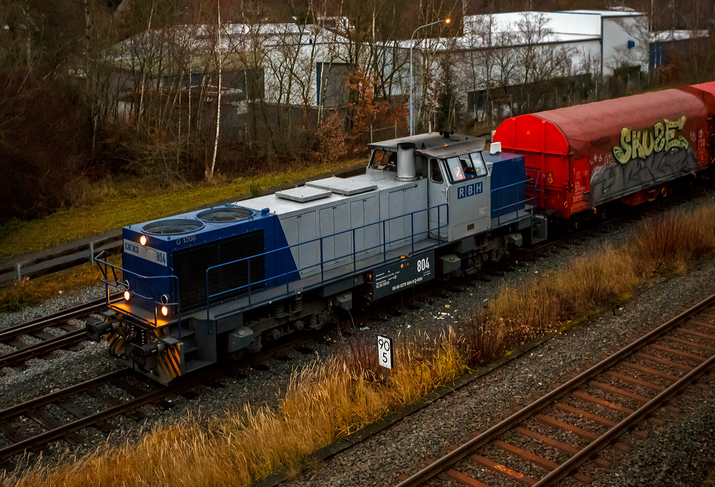 Aktuell besteht bei der KSW (Kreisbahn Siegen-Wittgenstein) immer noch Lokmangel, so ist zurzeit auch die RBH 804 / 275 804-9 (98 80 0275 804-9 D-RBH) eine MaK G 1206 der RBH - RAG Bahn und Hafen GmbH (Gladbeck), an die KSW vermietet. Hier fährt sie am 08 Dezember 2025 um 16:21 Uhr, beim allerletzten Licht des Tages, mit einem Übergabegüterzug (leere Wagen) auf dem Rbf der KSW in Herdorf (Betriebsstätte Freien Grunder Eisenbahn - NE 447) vor zur Gleisspeere (Anschlussstelle) zur Hellertalbahn. Die Helligkeit des Bildes täuscht etwas über die eigentlichen tatsächlichen Lichtverhältnisse hinweg.

Die MaK G 1206 wurde 1997 von der Siemens Schienenfahrzeugtechnik (SFT) in Kiel-Friedrichsort unter der Fabriknummer 1000903 gebaut und an die damalige RAG - Ruhrkohle AG, RAG Bahn- und Hafenbetriebe in Gladbeck als RAG 804 ausgeliefert, 2004 erfolgte die Umfirmierung in RAG Bahn und Hafen GmbH (RBH) und nach der Übernahme durch die DB Schenker Rail Deutschland AG (heute DB Cargo) erfolgte 2006 die Umfirmierung in RBH Logistics GmbH. 

Die SFT war die Nachfolgefirma der Krupp Verkehrstechnik GmbH bzw. der früheren Maschinenbau Kiel GmbH (MaK). Die SFT wurde am 1. Oktober 1998 von Siemens an die Vossloh AG verkauft, in Vossloh Schienenfahrzeugtechnik (VSFT) umbenannt und firmierte seit 23. April 2004 als Vossloh Locomotives GmbH. Die Vossloh-Lokomotiven aus Kiel trugen das Kürzel MaK in der Fahrzeug-Typenbezeichnung. Nach der Fokussierung von Vossloh auf den Bereich Schiene erfolgte 2019 ein weiterer Verkauf an die Chinesische CRRC Zhuzhou Locomotive Co., Ltd.

Die Lok (MaK G 1206):
Die MaK G 1206 ist im schweren Rangier- und Streckendienst einsetzbar. Sie wurde 1997 zum ersten Mal gebaut, erster Kunde war die RAG, somit diese (die 804) erst die Vierte der Serie. Bis 2016 wurden insgesamt 323 Fahrzeuge der Baureihe G 1206, in 5 verschiedenen Varianten, produziert. Sie wurden nach Deutschland, Österreich, der Schweiz, Luxemburg, Italien, Spanien, den Niederlanden und nach Frankreich verkauft worden. Allein 23 Lokomotiven fahren als BB 61000 bei der SNCF, davon sechs mit MTU-Motor (Die G 1206 gibt es wahlweise mit Caterpillar- oder MTU-Motor). 

Diese Lok der ersten Serie (von 11 Stück) hat einen wassergekühlten 90°V16- Zylinder-Viertakt-Dieselmotor mit Abgasturbolader und Ladeluftkühlung, sowie mit Direkteinspritzung, vom Typ MTU 16V 396 TC 14 des Herstellers MTU Friedrichshafen GmbH (MTU steht für Motoren- und Turbinen-Union). Der Motor hat ein Zylinderbohrung von 165 mm und einen Kolbenhub von185 mm, damit hat der Motor einen Hubraum von 63.3 Litern. Die Motorleistung beträgt 1.570 kW (2.135 PS) bei 1.800 U/min, solche Leitungen erbringe heute 12 Zylindermotoren. Das Strömungsgetriebe ist ein Voith L 5r4 zU2. Die Höchstgeschwindigkeit der Lok beträgt 80 km/h.

Die G 1206 nimmt im 3. Typenprogramm eine besondere Rolle ein. Sie ist nicht nur die größte und leistungsstärkste Mittelführerhaus-Lok als Schlusspunkt dieser Typenreihe, sondern hat mit ihrer Verbreitung in vielen Ländern Europas inzwischen eine gebaute Stückzahl erreicht wie noch keine unabhängig von Staatsbahnaufträgen entwickelte Standardlok aus Kiel vor ihr. Was 1997 als neue Lokgeneration für die Ruhrkohle AG begann, ist heute eine Lokomotive, die in acht europäischen Ländern bei zahlreichen Bahnen, darunter zwei Staatsbahnen, im Einsatz steht.

Mitte der 1990er Jahre entstand bei den Bahn- und Hafenbetrieben der Ruhrkohle AG Bedarf nach neuen Diesellokomotiven. Diese Maschinen sollten nicht nur im schweren internen Rangier- und Zustelldienst eingesetzt werden, sondern auch - die Möglichkeiten der Liberalisierung nutzend - mit schweren Kohlezügen auf Strecken der DB AG verkehren. Dafür benötigten die Lokomotiven eine entsprechend hohe Motorleistung und eine Höchstgeschwindigkeit, die einen freizügigen Einsatz auf DB-Gleisen zulässt.

Den Auftrag zur Entwicklung und Lieferung von elf dieser Lokomotiven bekam die damalige Siemens Schienenfahrzeugtechnik GmbH. Das Ergebnis war die G 1206, deren erstes Exemplar Ende Juli 1997 an die RAG ausgeliefert wurde. Sie ist aus der G 1205 entwickelt worden und unterscheidet sich von dieser hauptsächlich durch den Dieselmotor mit 16 statt 12 Zylindern und eine entsprechend dimensionierte Kühlanlage mit zwei Lüfterrädern. Um die größere Maschinenanlage auf der Lokomotive unterbringen zu können, ist die G 1206 fast zwei Meter länger als ihre Vorgängerin ausgeführt worden.

Die 5 verschiedenen Varianten unterscheiden sich vor allem an den verwendeten Motoren und Getrieben, wie folgt:
•	Motor MTU 16V 396 TC14 mit 1570 kW; Getriebe L 5r4 zU2; Hg 80 km/h
•	Motor MTU 16V 396 TC14 mit 1570 kW; Getriebe L 5r4 zseU2; Hg 60 / 100 km/h
•	Motor MTU 12V 4000 R20 mit 1500 kW; Getriebe L 5r4 zU2; Hg 80 km/h
•	Motor Caterpillar 3512B DI TA mit 1500 kW; Getriebe L 5r4 zU2; Hg 100 km/h
•	Motor MTU 12V 4000 R41 mit 1500 kW; Getriebe L 5r4 zseU2; Hg 60 / 100 km/h

TECHNISCHE DATEN der G 1206 der 1. Serie (RAG):
Spurweite: 1.435 mm (Normalspur)
Antriebart:  dieselhydraulisch
Achsfolge: B'B' 
Länge über Puffer: 14.700 mm
Drehzapfenabstand: 7.200 mm
Achsabstand im Drehgestell: 2.400 mm
größte Breite: 3.080 mm
Höhe über Schienenoberkante: 4.220 mm
Raddurchmesser: 1.000 mm (neu)
Motor: MTU 16V 396 TC14 
Leistung: 1.570 kW bei1.800 U/min
Strömungsgetriebe: Voith L 5r4 zU2
Anfahrzugkraft: 291 kN
Höchstgeschwindigkeit: 80 km/h
kleinster befahrbarer Gleisbogen: 60 m
Dienstgewicht: 88 t
Kraftstoffvorrat: 3.150 l
