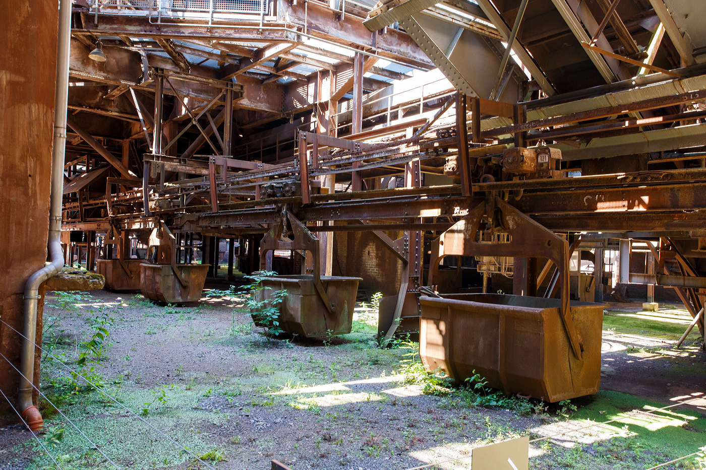 Auch eine Eisenbahn (Schwebebahn): Die H�ngebahn der V�lklinger H�tte am 01 Juli 2025, hier einer der selbstfahrenden H�ngebahnwagen unter der M�llerhalle. 

Die H�ngebahn ist eine Einschienenbahn bzw. Schwebebahn mit selbstfahrende H�ngebahnwagen, bei den Schr�gaufz�gen auf die Gichtb�hne wurden sie mit Seilz�gen unterst�tzt. 

Die H�ngebahn war seit 1911 in Betrieb. 1985 fuhren pro Stunde 240 beladene H�ngebahnwagen zur  Gicht . Sie liefen �ber die sogenannte Gichtb�hne in einer H�he von 27 Metern, die Rohstoffe (Eisenerz, Sinter, Schrott, Kalk und Koks) um die 6 Hoch�fen von dort zu beschicken. Die Gleisl�nge der H�ngebahn betrug �ber 6 km.

Der M�ller (Eisenerz, Sinter, Schrott und Kalk wurden in der M�llerhalle (s�d�stlich der Hoch�fen) in selbstfahrende H�ngebahnwagen verladen und �ber einen vierspurigen Schr�gaufzug auf die Gichtb�hne gezogen. Auf der Gichtb�hne laufen mehrere H�ngebahnspuren an den sechs Hoch�fen vorbei und wieder zur�ck zum Schr�gaufzug, mit kreisf�rmigen Abzweigschleifen um jeden Hochofen herum. Auf der Nordwestseite der Gichtb�hne l�uft ein kleinerer zweispuriger Schr�gaufzug hinunter zur Kokerei, um den Koks f�r die Hoch�fen zu f�rdern.

Die Gichtb�hne verbindet alle sechs Hoch�fen mit einer Ebene. Auf dieser Ebene werden die H�ngebahnwagen entlanggef�hrt. Die Gichtb�hne ist ca. 240 Meter lang und erstreckt sich in einer H�he von exakt 27 Meter.  Die Gichtb�hne hat Ihren Namen von dem Wort  Begichten , was das Bef�llen der Hoch�fen mit den Rohstoffen f�r die Eisenerzeugung bezeichnet. Eisenerz, Sinter, Zusatzstoffe, Koks, aber auch eisenhaltige Reststoffe aus der Industrie oder Schrott wurden in die Gicht, den Einf�lltrichter des Hochofens, gekippt. Die „Haube/obere Glocke“, der obere Verschluss des Hochofens, wurde angehoben, dann f�nf H�ngebahnwagen um den Trichter gefahren. Unterhalb der Haube befindet sich ein zweiter Verschluss, die „Gicht-Glocke“. Sie blieb geschlossen, um das Gichtgas aufzufangen. Das Material wurde auf die Glocke gekippt, dann die Haube abgesenkt. Erst wenn die Haube geschlossen war, wurde die Glocke nach unten gefahren: Das Material rutschte in den Hochofenschacht. Diese Verschlussart nannte man „Doppelglockenverschluss“, bei modernen Hoch�fen werden heute glockenlose Gichtverschl�sse verwendet. 
