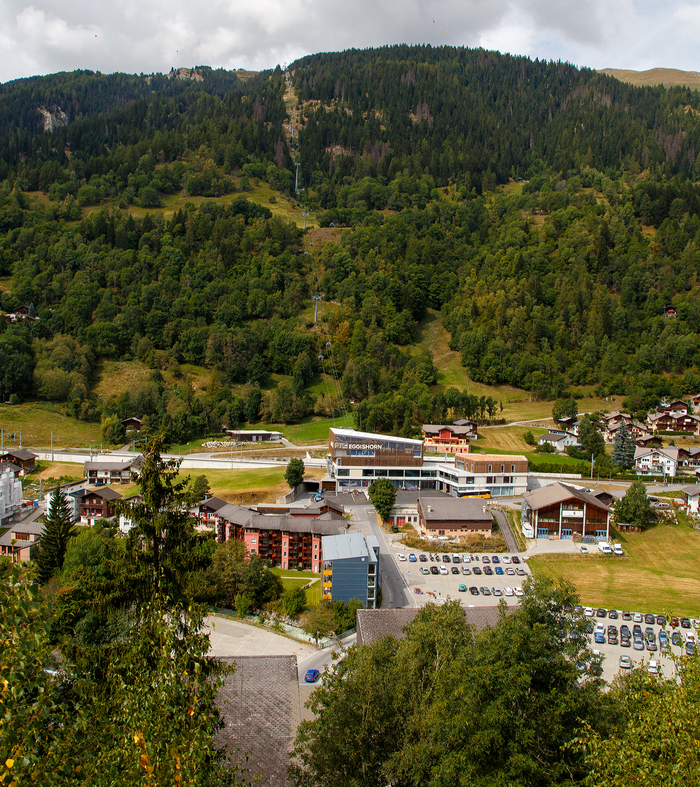 Blick auf den neuen �V-Hub Fiesch am 07 September 2021 aus einem MGB-Zug. Ein Jahrhundertbau in Fiesch, MGB Bahnhof (1.063 m �.M.) und Bus-Terminal mit dem direkten Zugang zur Talstation der neuen 10er-Kabinenbahn der Doppelmayr/Garaventa-Gruppe hinauf auf die Fiescheralp. 

Die neue 10er-Gondelbahn Fiesch - Fiescheralp / K�hboden, Baujahr 2019 von der Doppelmayr/Garaventa-Gruppe ist mit 7 m/s Fahrgeschwindigkeit die schnellste 10er-Gondelbahn der Schweiz. Sie ist eine sogenannte D-Line-Bahn der Doppelmayr/Garaventa-Gruppe mit Seillage�berwachung RPD. Als Betriebsmittel kommen die neuen CWA Kabinen Omega V zum Einsatz. Als Antrieb wurde der 867 kW starke Doppelmayr Sector Drive (DSD) verwendet, ein permanenterregter Synchronmotor, der mit modernster Frequenzumrichtertechnologie arbeitet und sich auch f�r steile Anlagen wie die hier in Fiesch sehr gut eignet.

Die neue Gondelbahn ersetzt zwei Pendelbahnen von Fiesch zur Fiescheralp, einmal mit 100er-Kabinen und einmal mit 34er-Kabinen. Die Talstation der hochmodernen Anlage befindet sich im neuen �V-Hub (Knotenpunkt �ffentlicher Verkehr) in Fiesch, wo direkter Anschluss an die Matterhorn-Gotthard-Bahn und das Postautonetz besteht.

Ein Ausbau der Bahn mit einer zweiten Sektion vom K�hboden Richtung Eggishorn als Ersatz f�r die Luftseilbahn Fiesch-Eggishorn ist angedacht und bereits genehmigt.

Immer mehr Skitouristen nutzen den �ffentlichen Verkehr f�r die Fahrt in den Wintersport. Voll diesem Trend entsprechend haben die Aletsch Bahnen, die Matterhorn-Gotthard-Bahn (MGB) und die Gemeinde Fiesch als Bauherren mit dem Architekten und Investor Hans Ritz die neue �V-Drehscheibe in Fiesch als Gemeinschaftsprojekt gebaut. Der neue Verkehrsknotenpunkt besteht aus der Talstation der neuen 10er-Kabinenbahn auf die Fiescheralp, einem komplett neuen Bahnhof der MGB und einem Postbus-Terminal. Durch die Integration und Verkn�pfung aller Verkehrstr�ger an einem Ort wurden kurze, schnelle und barrierefreie Umsteigwege geschaffen.

F�r das Projekt hat die MGB den Haltepunkt f�r ihre Z�ge vom bisherigen Bahnhofsgeb�ude 400 m nach Norden verlegt und in den �V-Hub integriert. Dadurch ist ein komplett neuer Bahnhof mit zwei Au�enbahnsteigen sowie einem 600 m langen neuen Doppelgleis f�r eine optimierte Kreuzungsm�glichkeit der Z�ge entstanden. Eine besondere Herausforderung bei der Gleisverlegung stellte die 36 m lange Bogenweiche mit einem Gewicht von 35 t dar. Sie wurde in drei Teilen angeliefert und dann mit einem Kran �ber die D�cher gehievt und eingebaut.

Die neue Kabinenbahn ersetzte die 100er-Pendelbahn Fiesch – Fiescheralp (K�hboden) von Garaventa aus dem Jahr 1974. F�r die neue Bergstation wurde die bestehende Bausubstanz der abgebauten Pendelbahn erweitert und f�r die Garagierung der Kabinen verwendet. Bei der Talstation wurden 760 m“ und bei der Bergstation 1.100 m� Kunststoff-Folie als Fassaden-verkleidung eingesetzt. Die Verkehrswege in den Stationen sind barrierefrei angelegt. Die Bahn ist mit der neuesten Kabinengeneration der Omega V von CWA ausgestattet, f�r den G�tertransport stehen zwei Transportkabinen zur Verf�gung.

Der �V-Hub Fiesch wurde nach einer �u�erst kurzen Bauzeit von 14 Monaten am 7. Dezember 2019 feierlich er�ffnet. Mit rund 24 Mio. CHF �bernahm die Aletsch Bahnen AG den Hauptanteil dieser Investition. Die MGB beteiligte sich mit 17,6 Mio. CHF, der Architekt Hans Ritz investierte 6 Mio. Das Vorzeigeprojekt �V-Hub Fiesch macht eindrucksvoll deutlich, was durch Kooperation von verschiedenen Partnern erreicht werden kann.

TECHNISCHE DATEN der Seilbahn:
Hersteller:  Garaventa  (Doppelmayr/Garaventa-Gruppe)
Typ: 10er Gondelbahn (Ein-Seil-Umlaufbahn)
Baujahr: 2019
H�he der Talstation:1.083 m �. M.
H�he der Bergstation: 2.227 m �. M.
H�hendifferenz: 1.144 m
Abstand zwischen Tal- und Bergstation: 2.698 m
Anzahl der St�tzen: 15
F�rderseildurchmesser: 56 mm
Antriebsstation: Berg (Spanneinrichtung Tal)
Motorleistung: 867 kW (im Betrieb) / 1.124 kW (beim Anfahren)
Geschwindigkeit:  7 Meter pro Sekunde
Effektive Fahrzeit: 7:48 Minuten
F�rderleistung: 1.880 Personen pro Stunde
Kabinenanzahl: 48 und 2 Transport-/G�terkabinen
Kabinentyp: CWA Omega V