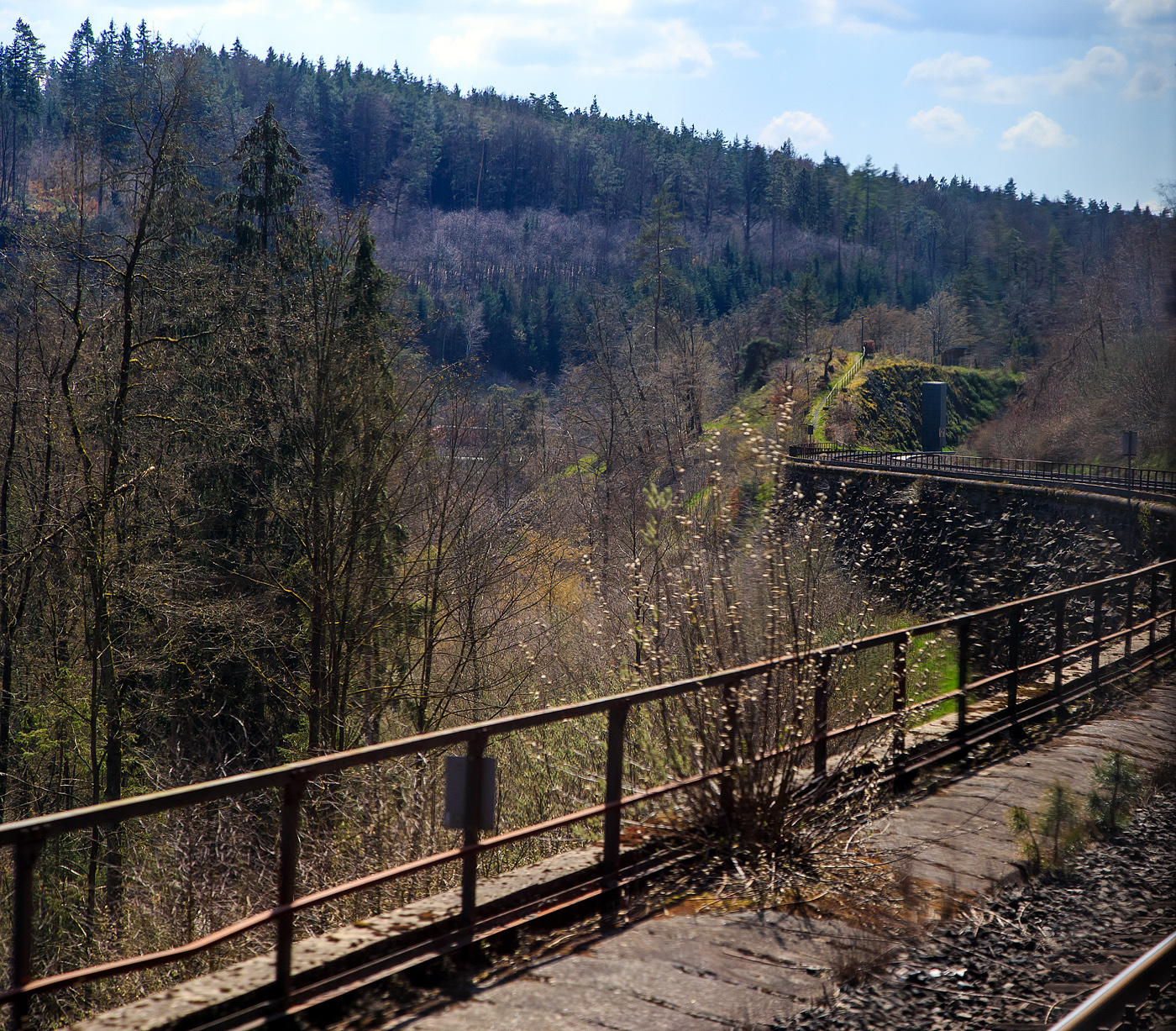 Blick aus dem Zug heraus...
Nun fahren wir am 21.04.2023, mit dem RE 30 von Hof nach Bamberg, die ber�hmte Schiefe Ebene von Marktschorgast nach Neuenmarkt-Wirsberg hinab. 

Die Schiefe Ebene ist ein Abschnitt der Ludwig-S�d-Nord-Bahn von Bamberg nach Hof (Saale), eine Steilstrecke mit der maximalen Neigung von 25 ‰ (1:40).

Vorne bei Kilometer 79,8 der Strecke (zwei Kilometer unterhalb von Marktschorgast) sieht man einen der beiden Pfeiler (beiderseits der Strecke), der andere steht recht dem gegen�ber (nicht im Bild), diese bestehen jeweils aus neun Betonsegmenten uns sind eine Fallk�rpersperre“. Diese wurden 1983, in der Zeit des Kalten Kriegs, gebaut. Im Ernstfall w�re in einem darunterliegenden Hohlraum befindlicher Sprengstoff zur Explosion gebracht worden, die Betonsegmente w�ren auf die Gleise gest�rzt. Damit hoffte man, anr�ckende Truppen des Warschauer Pakts aufhalten zu k�nnen. Diese Anlage steht heute unter Denkmalschutz.

Die Schiefe Ebene liegt im Landkreis Kulmbach, im Regierungsbezirk Oberfranken in Bayern. Sie beginnt �stlich des Bahnhofs Neuenmarkt-Wirsberg und endet im Bahnhof Marktschorgast. Die Rampe �berwindet auf dem Weg vom Maintal zur Rhein-Elbe-Wasserscheide auf der M�nchberger Hochfl�che auf 6,8 Kilometern 157,7 H�henmeter mit einer Steigung von bis zu 25 ‰. Wie die gesamte Ludwig-S�d-Nord-Bahn wurde auch die Schiefe Ebene mit einem Unterbau f�r eine zweigleisige Strecke errichtet, zun�chst aber nur ein Gleis verlegt. Erst 1871 wurden die Gelder f�r das zweite Gleis zwischen Untersteinach und Oberkotzau bewilligt. Wegen der zahlreichen St�tzmauern, Einschnitte und Steind�mme gilt sie als technische Meisterleistung ihrer Zeit. Sie ist heute ein Kulturdenkmal aufgrund des Bayerischen Denkmalschutzgesetzes.

Die Strecke ist heute durchgehend zweigleisig ausgebaut und nicht elektrifiziert. Auf dem sich an die stark geneigte Strecke anschlie�enden Abschnitt zwischen Marktschorgast und Stammbach wurde das zweite Streckengleis inzwischen abgebaut. �stlich des Bahnhofs Neuenmarkt-Wirsberg wurde f�r den schnellen Neigetechnikverkehr 2001 die sogenannte Schl�mener Kurve er�ffnet, mit der dieser Bahnhof auf dem Weg von Bayreuth nach Hof umfahren werden kann.

Die Strecke war zu Dampflokzeiten eine betriebliche Herausforderung. Viele Z�ge mussten durch Schiebelokomotiven oder eine zweite, vorgespannte Lokomotive verst�rkt werden. Sie waren im Bahnbetriebswerk Neuenmarkt-Wirsberg in Neuenmarkt stationiert. Darunter waren Lokomotiven der Baureihe 95, aber auch Lokomotiven der Baureihen 57 und 50. Anfang der 1970er-Jahre war die Strecke einer der letzten Einsatzorte der Baureihe 01 der Deutschen Bundesbahn.
