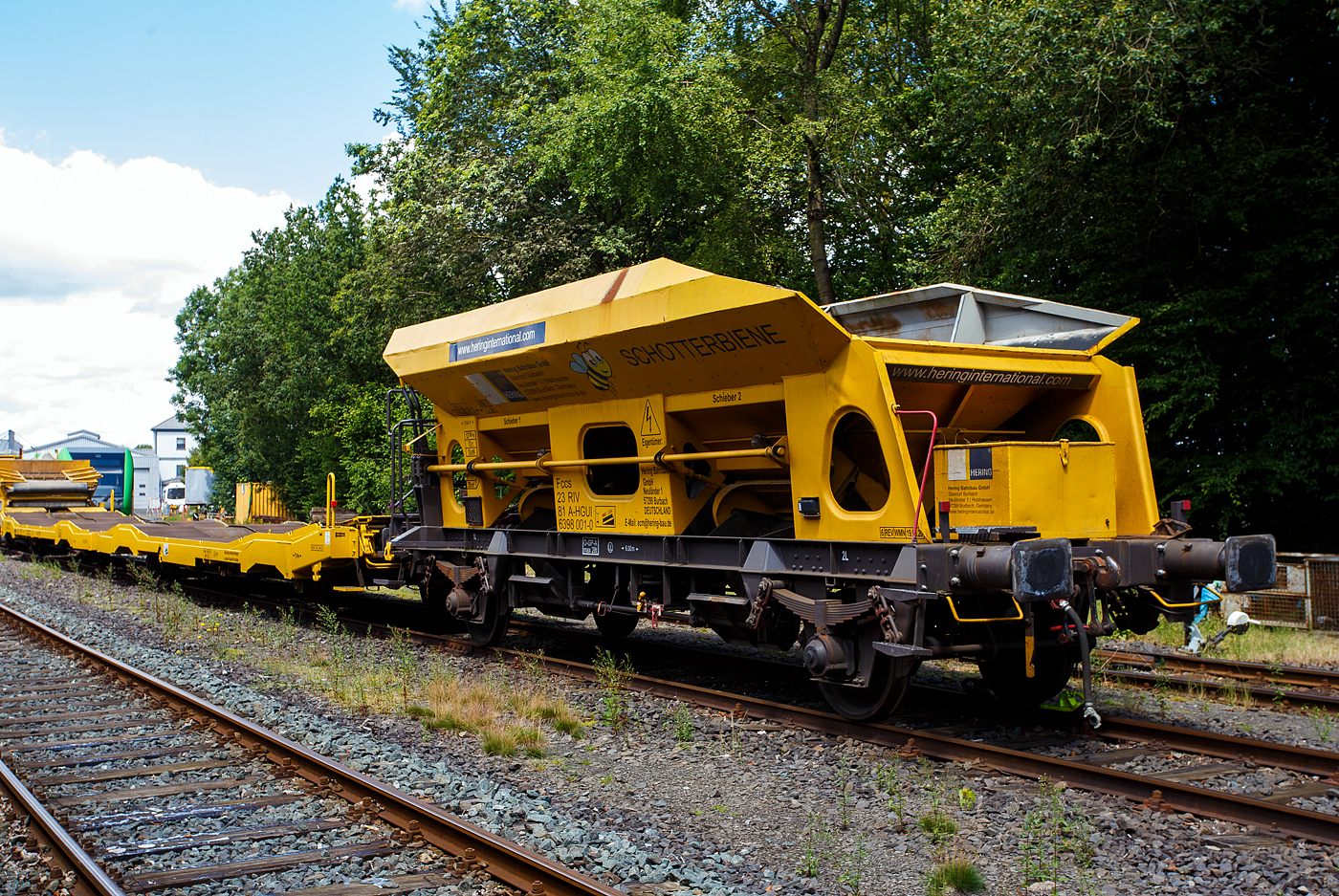 Das Allroundtalent die „Schotterbiene“ , der Schüttgutwagen mit dosierbarer Schwerkraftentladung, 23 81 6398 001-0 A-HGUI, „Schotterbiene“, der Gattung Fccs, der HERING Bahnbau GmbH (Burbach), ist am 07 Juli 2024 auf dem Betriebshof der Westerwaldbahn (WEBA) auf der Bindweide bei Steinebach/Sieg abgestellt.

Der Wagen wurde 1981 von der SGP - Simmering-Graz-Pauker AG im Werk Graz unter der Fabriknummer 76129 gebaut. Zusätzlich war noch „ÖBB.573.3.945“ über dem Fabrikschild eingeschlagen, so war der Ursprung des Wagens wohl die ÖBB. Hier in der Werkstatt der Westerwaldbahn wurde der Wagen im Dezember 2020 abgeändert und zur „Schotterbiene“ umgebaut. An der Bedienbühne gegenüberliegende Stirnseite wurde oben großzügig ein Stück entfernt. So ist es nun möglich den Wagen mittels einem Förderband einer Materialförder- und Siloeinheiten MFS oder Beladestation BLS 2000 zu beladen. Warum die HERING Bahnbau in Österreich eingestellt hat ist mir nicht ganz klar, vielleicht ist es dort einfacher oder weil der Wagen aus Österreich stammt.

Bedeutung der Gattung Fccs:
F = Offener Güterwagen in Sonderbauart
cc = dosierbare Schwerkraftentladung, wahlweise zweiseitig, tiefliegend (kleiner 70 cm über Schienenoberkante, und somit keine Entladung auf Fördereinrichtungen möglich)
s = zugelassen für Züge bis 100 km/h

Mit der „Schotterbiene“ von HERING in Verbindung mit einer MFS-Einheit kann Schotter als Füllschotter, Oberschotter oder Stopfschotter im Gleis sicher, effizient und leise eingebaut werden. Dabei wird im Schottervorgang der zuvor in die MFS-Wagen aufgebunkerte Schotter in den Schottertrichter des Trichterwagens vor Kopf gefördert.

Bei den offenen Selbstentladewagen hat der Laderaum die Form zweier nebeneinander stehenden Trichtern der Entladevorgang erfolgt über die Schwerkraft des Ladegutes. Zwei Auslauföffnungen mit Drehscheiben ermöglicht die restlose Entladung des Wagens. Die Menge ist regulierbar, so wie es zum Beispiel beim Einschottern erforderlich ist. Über vier unabhängig voneinander verstellbare Lenkbleche wird der Schotter danach ins Arbeitsgleis gelenkt und abgelassen. Sowohl zwischen den Schienen im Innenbereich als auch außerhalb der Schienen kann Schotter, auch gleichzeitig, abgelassen werden. Die Bedienung des Wagens erfolgt manuell, von der stirnseitigen Bedienungsbühne. Die Drehschieber können einzeln geöffnet, reguliert und geschlossen werden. Der Arbeitsbereich ist gut ausgeleuchtet. Beide Trichter sind innen mit einem Gummibelag ausgekleidet, was eine maximale reduzierte Geräuschemission beim Ablassen des Schotters ermöglicht. Selbstverständlich kann die Schotterbiene auch als üblicher Schotterwagen verwendet werden!

TECHNISCHE DATEN:
Hersteller: SGP - Simmering-Graz-Pauker AG im Werk Graz
Baujahr/Fabriknummer: 1981 / 76129
Umbau: 2020 durch WEBA
Gattung: Fccs  Schotterbiene 
Spurweite: 1.435 mm (Normalspur)
Achsanzahl: 2
Länge über Puffer: 9.640 mm
Achsabstand: 6.000 mm
Zulade Volumen: 20 m³
Eigengewicht: 12.700 kg
Max. Zuladung: 27,0 t (ab Streckenklasse C) / ÖBB 28,5 t bei D
Höchstgeschwindigkeit: 100 km/h
Bremse: O – GP – A (max. 28 t) (LL)
Intern. Verwendungsfähigkeit: RIV

Quelle: HERING Bahnbau (der Verwendungsart), Daten laut Anschriften