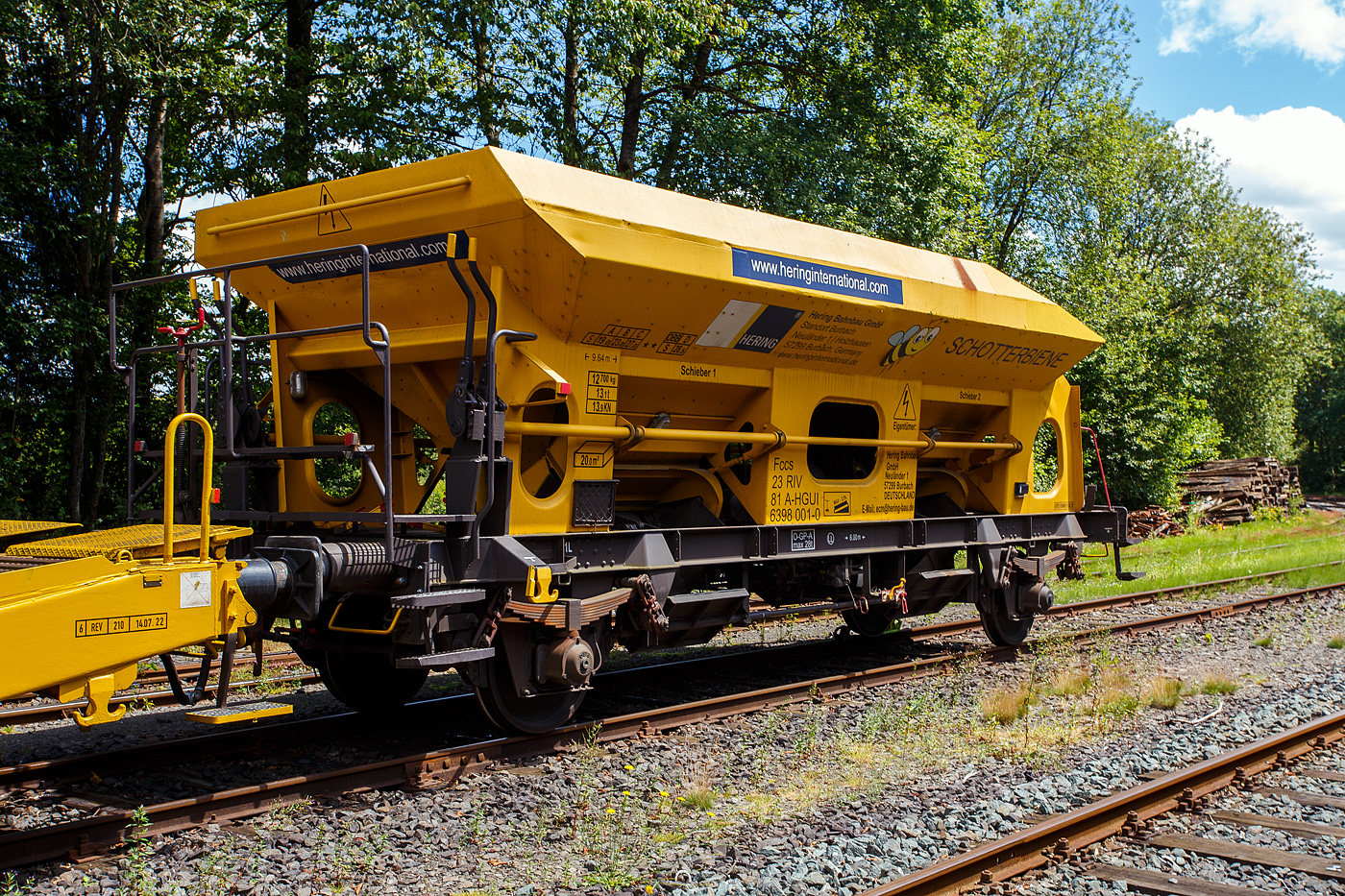 Das Allroundtalent die „Schotterbiene“ , der Schüttgutwagen mit dosierbarer Schwerkraftentladung, 23 81 6398 001-0 A-HGUI, „Schotterbiene“, der Gattung Fccs, der HERING Bahnbau GmbH (Burbach), ist am 07 Juli 2024 auf dem Betriebshof der Westerwaldbahn (WEBA) auf der Bindweide bei Steinebach/Sieg abgestellt.

Der Wagen wurde 1981 von der SGP - Simmering-Graz-Pauker AG im Werk Graz unter der Fabriknummer 76129 gebaut. Zusätzlich war noch „ÖBB.573.3.945“ über dem Fabrikschild eingeschlagen, so war der Ursprung des Wagens wohl die ÖBB. Hier in der Werkstatt der Westerwaldbahn wurde der Wagen im Dezember 2020 abgeändert und zur „Schotterbiene“ umgebaut. An der Bedienbühne gegenüberliegende Stirnseite wurde oben großzügig ein Stück entfernt. So ist es nun möglich den Wagen mittels einem Förderband einer Materialförder- und Siloeinheiten MFS oder Beladestation BLS 2000 zu beladen. Warum die HERING Bahnbau in Österreich eingestellt hat ist mir nicht ganz klar, vielleicht ist es dort einfacher oder weil der Wagen aus Österreich stammt.

Bedeutung der Gattung Fccs:
F = Offener Güterwagen in Sonderbauart
cc = dosierbare Schwerkraftentladung, wahlweise zweiseitig, tiefliegend (kleiner 70 cm über Schienenoberkante, und somit keine Entladung auf Fördereinrichtungen möglich)
s = zugelassen für Züge bis 100 km/h

Mit der „Schotterbiene“ von HERING in Verbindung mit einer MFS-Einheit kann Schotter als Füllschotter, Oberschotter oder Stopfschotter im Gleis sicher, effizient und leise eingebaut werden. Dabei wird im Schottervorgang der zuvor in die MFS-Wagen aufgebunkerte Schotter in den Schottertrichter des Trichterwagens vor Kopf gefördert.

Bei den offenen Selbstentladewagen hat der Laderaum die Form zweier nebeneinander stehenden Trichtern der Entladevorgang erfolgt über die Schwerkraft des Ladegutes. Zwei Auslauföffnungen mit Drehscheiben ermöglicht die restlose Entladung des Wagens. Die Menge ist regulierbar, so wie es zum Beispiel beim Einschottern erforderlich ist. Über vier unabhängig voneinander verstellbare Lenkbleche wird der Schotter danach ins Arbeitsgleis gelenkt und abgelassen. Sowohl zwischen den Schienen im Innenbereich als auch außerhalb der Schienen kann Schotter, auch gleichzeitig, abgelassen werden. Die Bedienung des Wagens erfolgt manuell, von der stirnseitigen Bedienungsbühne. Die Drehschieber können einzeln geöffnet, reguliert und geschlossen werden. Der Arbeitsbereich ist gut ausgeleuchtet. Beide Trichter sind innen mit einem Gummibelag ausgekleidet, was eine maximale reduzierte Geräuschemission beim Ablassen des Schotters ermöglicht. Selbstverständlich kann die Schotterbiene auch als üblicher Schotterwagen verwendet werden!

TECHNISCHE DATEN:
Hersteller: SGP - Simmering-Graz-Pauker AG im Werk Graz
Baujahr/Fabriknummer: 1981 / 76129
Umbau: 2020 durch WEBA
Gattung: Fccs  Schotterbiene 
Spurweite: 1.435 mm (Normalspur)
Achsanzahl: 2
Länge über Puffer: 9.640 mm
Achsabstand: 6.000 mm
Zulade Volumen: 20 m³
Eigengewicht: 12.700 kg
Max. Zuladung: 27,0 t (ab Streckenklasse C) / ÖBB 28,5 t bei D
Höchstgeschwindigkeit: 100 km/h
Bremse: O – GP – A (max. 28 t) (LL)
Intern. Verwendungsfähigkeit: RIV

Quelle: HERING Bahnbau (der Verwendungsart), Daten laut Anschriften