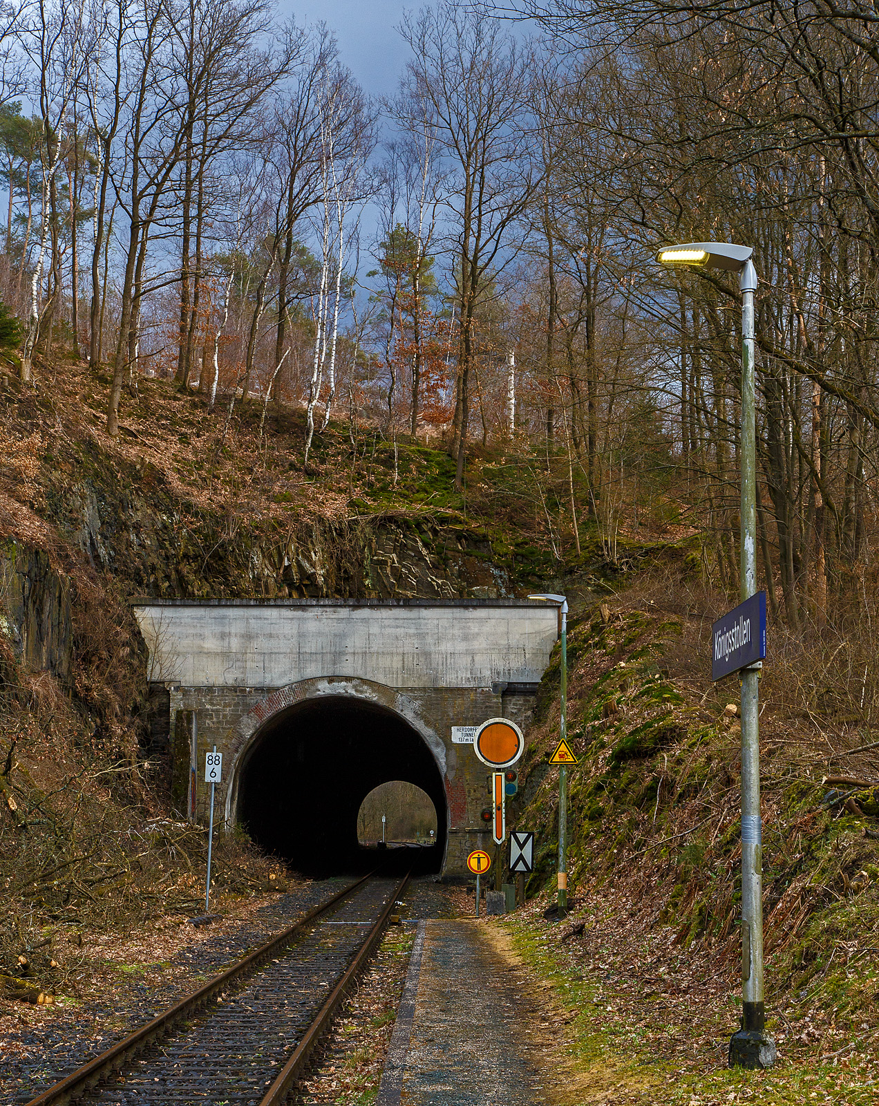 Der 137 m lange Herdorfer Tunnel hier am 18.03.2023 vom Hp K�nigsstollen gesehen. 

Davor das Vorsignal (f�rs Einfahrtsignal Bf Herdorf) Vr 0 - Halt erwarten! Die gelbe Scheibe mit wei�em Rand und gelbe Pfeil zeigt nach unten. Nachts leuchten auch die beiden gelben Lampen. Vor dem Signal die Ne 2 – Vorsignaltafel. Die Vorsignaltafel zeigt dem Lokf�hrer, da� das Signal, vor dem sie steht, Vorsignalfunktion hat, d.h., den Begriff eines nachfolgenden Hauptsignales vorank�ndigt. 
