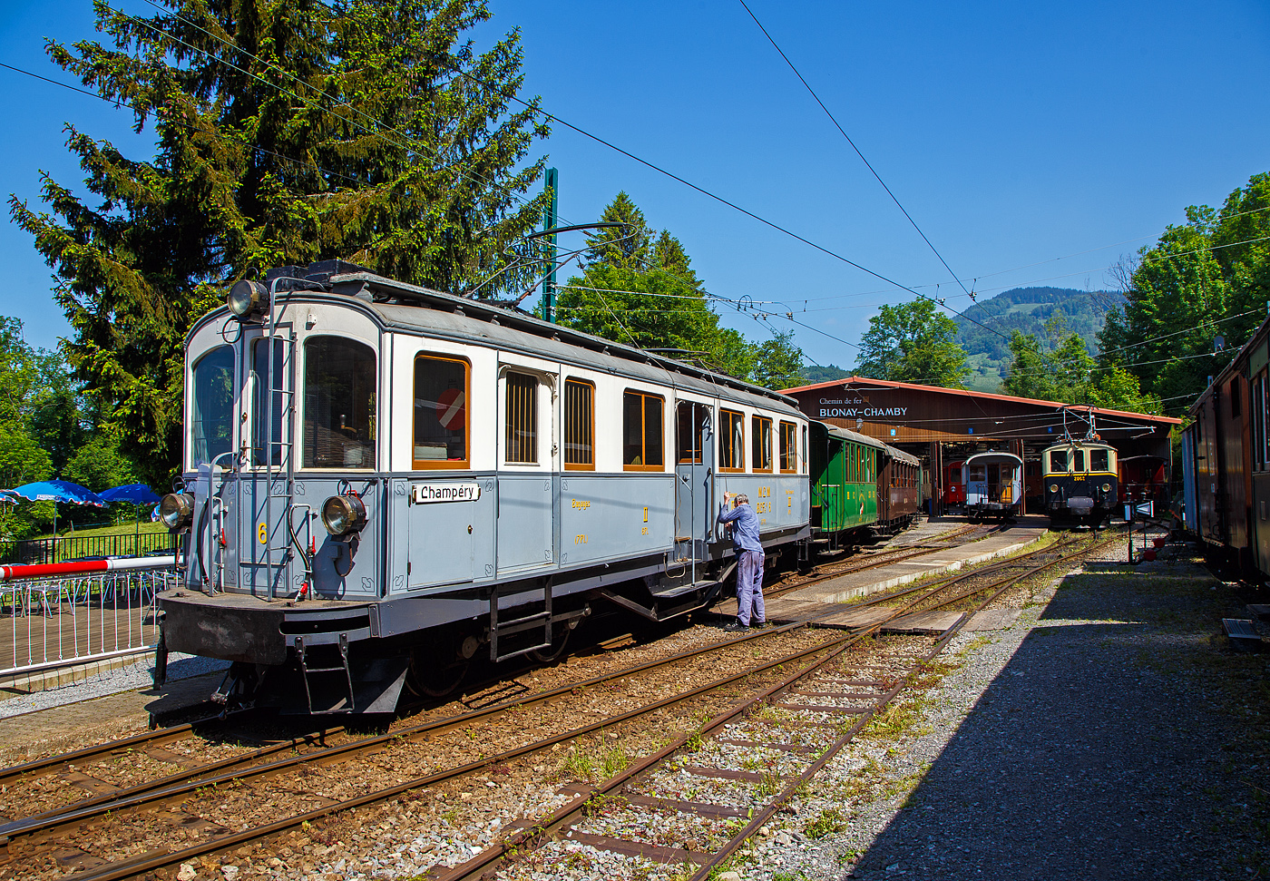Der 2./3. Klasse elektrische Personentriebwagen mit Gep�ckabteil f�r den gemischten Zahnrad- und Adh�sionsbetrieb ex MCM BCFeh 4/4 6 (nach heutiger Bezeichnung eigentlich BCDeh 4/4) der Museumsbahn Blonay–Chamby steht am 27 Mai 2023 im Museum Chaulin mit einem Zug zur Abfahrt bereit. Der Lokf�hrer hat gerade noch den Lyra-Stromabnehmer mit der Schnur abgesenkt. Beim Heben und Senken der Stromabnehmer muss hier noch Hand angelegt werden.

Die BCFeh 4/4 1–3 sind elektrische Personentriebwagen mit Gep�ckabteil f�r den gemischten Zahnrad- und Adh�sionsbetrieb, die 1908 zur Betriebser�ffnung der meterspurigen Monthey-Champ�ry-Morgins-Bahn (MCM) beschafft wurden. Im Jahr 1946 entstand durch Fusion der MCM und der Aigle–Ollon–Monthey-Bahn (AOM) die Aigle-Ollon-Monthey-Champ�ry-Bahn (AOMC), die wiederum fusionierte 1999 mit der AL, ASD und BVB zur der heutigen Transports Publics du Chablais (TPC). 

Die Triebwagen der MCM, sp�teren AOMC hatten das Zahnstangensystem Strub, 2016 wurden auf der Strecke Aigle–Ollon–Monthey–Champ�ry die Zahnstangenabschnitte erneuert und das bisherige Zahnstangensystem Strub durch das Zahnstangensystem Abt ersetzt. Fahrleitungsspannung wurde auf 1500 Volt erh�ht. Seither besitzen die ab Aigle verkehrenden Strecken der TPC das gleiche Zahnstangensystem und die gleiche Spannung. So k�nnen die modernen Stadler Beh 2/6 SURF auf allen drei Strecken verkehren.

Bei dem hier gezeigten MCM BCFeh 4/4 6 (sp�ter ABFeh 4/4, ab 1966 ABDeh 4/4) handelt es sich um einen 1909, bereits ein Jahr nach der Betriebser�ffnung, erfolgten Gelegenheitskauf eines weitgehend baugleichen Triebwagen, der urspr�nglich f�r die Stresa-Mottarone-Bahn (FSM) in Italien bestimmt war. Die f�r das Zahnstangensystem Strub ausgelegten Triebwagen wurden von der Schweizerischen Industrie-Gesellschaft in Neuhausen am Rheinfall erbaut, wobei die elektrische Ausr�stung von der Elektrizit�tsgesellschaft Alioth in M�nchenstein und die Drehgestelle einschl. des Zahnradantriebs von der Schweizerischen Lokomotiv- und Maschinenfabrik (SLM) in Winterthur stammen. Der Triebwagen BCFeh 4/4 6 ist als einziger bei der Museumsbahn Blonay–Chamby (BC) in der Westschweiz erhalten geblieben. die Triebwagen 1 bis 3 wurden 1954 au�er Betrieb gesetzt und dann abgebrochen.

Die Triebwagen charakterisierten sich bei der Ablieferung durch die beiden Drehgestelle mit Stangenantrieb, einen Mitteleinstieg und Holzrahmenfenster. Ebenso typisch ist f�r diese Triebwagen, dass die Mittelpufferkupplung mit unten liegender einfacher Schraubenkupplung in einer Verl�ngerung des Untergestells integriert ist. Ansonsten haben sie die damals �bliche Bauweise mit einem Untergestell und einem verblechten Holzaufbau.

In den beiden Drehgestellen sind jeweils ein Motor f�r den Adh�sions- und einer f�r den Zahnradantrieb untergebracht. Ersterer treibt eine der beiden Achsen �ber ein einfach �bersetztes Getriebe an, von der aus wird die andere Achse �ber au�erhalb des Drehgestellrahmens liegende Kuppelstangen angetrieben. Der andere Motor treibt das auf der zweiten Achse lose aufgesetzte Zahnrad �ber eine doppelte �bersetzung an.

Im Adh�sionsbetrieb sind nur die beiden daf�r vorgesehenen Motoren in Betrieb. Beim Einsatz im Zahnradbetrieb arbeiten alle vier Motoren, wobei die Adh�sionsmotoren hier der deutlich langsameren Fahrgeschwindigkeit wegen nur in Serienschaltung eingesetzt werden k�nnen. Demzufolge stehen den Triebwagen Nummer 1–3 im Adh�sionsbetrieb 2 � 75 PS, insgesamt nur 150 PS zur Verf�gung. Im Zahnradbetrieb sind es 2 � 75 PS f�r die beiden Motoren des Zahnradantriebes und die H�lfte von 2 � 75 PS f�r die Motoren des Adh�sionsbetrieb, insgesamt 225 PS. 

Beim Triebwagen Nummer 6 stehen nach den gleichen rechnerischen Grunds�tzen im Adh�sionsbetrieb 2 � 95 PS = 190 PS zur Verf�gung, im Zahnstangenbetrieb 285 PS. Alle vier Triebwagen hatten/haben eine Druckluft- und Widerstandsbremse. Zwei unabh�ngig auf die Zahnr�der und die Treibr�der des Adh�sionsbetriebs wirkende Handbremsen erg�nzten die Bremsausr�stung.

Einsatzgebiete
Die Triebwagen verkehrten fast ausschlie�lich auf der mit mehreren Zahnstangenabschnitten versehenen Bahnstrecke Monthey–Champ�ry. Einzig der Triebwagen Nummer 6 wurde nach der umfassenden Erneuerung des Fahrzeugparkes 1954 in der Funktion als Diensttriebwagen auch auf der Bahnstrecke Aigle–Ollon–Monthey eingesetzt. Die Fahrzeuge besa�en w�hrend der ganzen Betriebsdauer einen hellgrau/wei�en Anstrich. Nur der Triebwagen 6 war sp�testens ab 1954 bis 1975 in gr�n/creme lackiert.

Verbleib
Nach der Ablieferung der neuen Triebwagen f�r gemischten Adh�sions- und Zahnradbetrieb BDeh 4/4 501 bis 504 wurden die Triebwagen 1 bis 3 1954 au�er Betrieb gesetzt und dann abgebrochen. Erhalten geblieben ist der etwas st�rkere und ein Jahr j�ngere Triebwagen 6. Dies obschon er 1971 auch au�er Betrieb gesetzt wurde, wurde dieser 1975 wieder in Betrieb genommen. Nachdem er durch die Museumsbahn Blonay–Chamby wieder in den urspr�nglichen Farben lackiert worden ist, wurde er nach einer letzten Fahrt auf seiner urspr�nglichen Bahnstrecke im Jahre 1976, als Schenkung �bernommen. Anl�sslich der 100-Jahres-Feier der Aigle–Ollon–Monthey–Champ�ry-Bahn im Jahre 2008 kehrte er auf seine Stammstrecke zur�ck und wurde bei den Transports Publics du Chablais (TPC) in En Ch�lex zusammen mit dem Personenwagen BC2 10 von 1908 aufgearbeitet.

TECHNSCHE DATEN des BCFeh 4/4 Nr. 6 (1 bis 3 weichen leicht ab):
Baujahr: 1909
Spurweite: 1.000 mm
Achsfolge: Bz’Bz’
Zahnradsystem: 	Strub
L�nge �ber Puffer : 13.600 mm
L�nge Lokkasten: 12.600 mm 
Breite: 2.500 mm
Drehzapfenabstand: 7.500 mm
Achsstand im Drehgestell: 1.800 mm
Triebraddurchmesser: 914 mm (neu)
Zahnradteilkreis-�: 732 mm
Dienstgewicht: 30,2 t (27,3 t Nr. 1 – 3)
Leistung (Adh�sion): 2 � 95 PS = 190 PS (2 x 70 kW = 140 kW)
Leistung (Zahnradbetrieb): 285 PS  (210 kW)
Getriebe�bersetzungen: 1:5.62 (Adh�sion)  / 1:9.77 (Zahnrad)
Zul. H�chstgeschwindigkeit (Adh�sion): 25 km/h 
Zul. H�chstgeschwindigkeit (Zahnradbetrieb): 15 km/h (Talfahrt 10 km/h)
Stromsystem: 850 V DC (Gleichstrom)
Stromabnehmer: 2 Lyra-Stromabnehmer, Bet�tigung �ber Schnur
Max. Neigung : 135 ‰
Sitzpl�tze: 6 (2. Klasse), 24 (3. Klasse) sowie 7 Klappsitze im Gep�ckabteil
Gep�ckabteilgr��e: 4.8 m�
Max. Gep�ck Zuladegewicht: 0,7 t

Quellen: BC und Wikipedia
