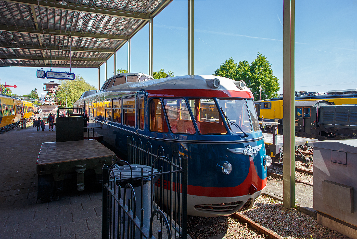 Der Dieseltriebwagen NS 20 „de Kameel“ / das Kamel bzw. VIP-Car, ein Dieseltriebwagen – Einzelstück und ehemaliger Inspektionstriebwagen, am 29 April 2025 im Het Spoorwegmuseum (bis 2005 NSM - Nederlands Spoorwegmuseum / Niederländischen Eisenbahnmuseum) in Utrecht-Maliebaan im Bahnhof Maliebaan. Ein sehr schönes Eisenbahnmuseum das zum Verweilen einlädt, leider fehlte uns dieses Mal die Zeit dafür. 

Der dieselelektrische Triebwagen wurde 1954 von Allan & Co´s Koninklijke Nederlandsche Fabrieken van Meubelen en Spoorwegmaterieel N.V. (Königlich Niederländische Möbel- und Eisenbahnausrüstungsfabrik) in Rotterdam gebaut und als Inspektionstriebwagen NS 20 an die NS - Nederlandse Spoorwegen (Niederländische Staatsbahnen) geliefert. Er diente für Inspektionsfahrten des NS-Vorstands.

Er technisch von den Triebwagen vom Typ DE 1 - Plan X „Blauwe Engel“ abgeleitet, nur der große Unterschied sind die Ausbuchtungen mit den Führerständen am Dach, die im bald den Spitznamen „Kamel“ einbrachten. 

1972 stand für den NS 20 eine Generalüberholung an. Zuvor durfte der Triebwagen in Toppop-Lackierung für die Musiksendung „Toppop“ durch die Niederlande touren. Im Rahmen der Überholung wurde der Triebwagen in Gelb umlackiert. Nach dieser Überholung stand der als „VIP-Wagen“ bekannte Triebwagen für Gruppenreisen zur Verfügung. Am 31. Januar 1991 fuhr der Wagen zum letzten Mal für die Öffentlichkeit, und am 8. März erfolgte die letzte Fahrt für die NS-Leitung. Anfang April fuhr die NS 20 mit eigenem Antrieb zur Lijnwerkplaats (Werkstatt) in Zwolle, um seine Generatoren als Ersatzteile zu spenden und sie der Reserve für die (inzwischen umgebauten und gelb lackierten) Blauwe-Engel-Züge hinzuzufügen. Diese Züge litten damals unter einem Mangel an Ersatzteilen.

Von diesem Zeitpunkt an war der NS 20 nicht mehr betriebsbereit und wurde zusammen mit einzelnen Dieselloks von der Lokomotive NS 2205 zum Eisenbahnmuseum transportiert und dort ausgestellt.

2005 wurde erneut ein Inspektionsfahrzeug benötigt, sodass eine Generalüberholung eingeleitet wurde, um das „Kamel“ wieder fahrbereit zu machen. Aus den ausgemusterten Plan X „Blaue Engel“ wurden Generatoren ausgebaut, zwei davon ausgewählt und nach Überholung wieder in den NS 20 eingebaut. Die Niederspannungsversorgung und die Küchenausstattung wurden in mehreren Bereichen für den modernen Einsatz modifiziert. Der in seiner originalen blauen Lackierung lackierte Triebwagen kehrte am 15. März 2008 mit eigener Kraft ins Museum zurück und wird dort betriebsbereit aufbewahrt.

Technik:
Auch die Frontpartie des Triebwagens anders als bei den DE 1 und 2. Das „Kamel“ ist 25.555 mm lang, somit fast 1,5 m kürzer als ein DE 1. Er hat eine Höhe von 4,145 Metern und eine Breite von 2,780 Metern. Das Gewicht des Fahrzeugs beträgt 58 Tonnen.

Der dieselelektrische Triebwagen verfügt über zwei Drehgestelle, die jeweils von zwei Elektromotoren angetrieben werden. Die elektrische Energie wird von zwei wassergekühlte AEC-6-Zylinder-Unterfur-Dieselmotoren Typ A220 (geliefert von Kemper & Van Twist), mit einer max. Leistung 132 kW (180 PS), Dauerleistung 107 kW (145 PS) mit gekoppeltem einem Generator von Smit vom Type G40/35 (147 kW) erzeugt, die jeweils zwei Heemaf-Fahrmotoren, Typ TM 51, antreiben. Die Dieselmotoren der DE 1 und 2 - Plan X „Blaue Engel“ hingegen haben durch einen Turbolader von Brown-Boveri eine höhere Dauerleistung von 165 kW (225 PS). Die maximale Drehzahl des Dieselmotors beträgt 1.250 U/min. Der Dieselmotor mit Generator, sowie Kompressor sind jeweils unter der Karosserie des Triebwagens montiert.

Der Triebwagen ist mit vier Heemaf-Fahrmotoren vom Typ TM 51 mit einer maximalen Leistung von 66 kW, 337 A bei 1.130 U/min und einer Dauerleistung von 49 kW, 248 A und 1.290 U/min ausgestattet. Die Energie liefert jeweils ein vom Dieselmotor angetriebener Generator für je 2 Motore. Kompressor und Hilfsgenerator werden über Keilriemen angetrieben. Diese befinden sich auf der anderen Seite des Hauptgenerators.

Der Radstand der Drehgestelle beträgt 3,0 Meter, der Mittenabstand zwischen den Drehgestellen 18,35 Meter. Der NS 20 ist für eine Höchstgeschwindigkeit von 125 km/h zugelassen. Der Raddurchmesser der Triebdrehgestelle, ist jedoch größer als bei den anderen Plan X-Garnituren, nämlich 1.250 mm. Die Drehgestelle sind jeweils mit zwei Heemaf-Fahrmotoren vom Typ TM 51 ausgestattet. Die Achsfolge ist somit Bo'Bo'. 

TECHNISCHE DATEN des DE 1 – 20 „de Kameel“:
Gebaute Anzahl: 1
Nummerierung: 20
Typ: dieselelektrischer Triebzug
Baujahr: 1954
Hersteller: Allan, Rotterdam
Spurweite: 1.435 mm (Normalspur)
Achsfolge: Bo'Bo' (4 Achsen in 2 Drehgestellen)
Länge über Kupplung: 25.550 mm
Drehzapfenabstand:
Achsanstand im Drehgestell: 3.000 mm
Breite: 2.780 mm
Höhe: 4.145 mm
Treibraddurchmesser: 1.250 mm (neu) 
Dieselmotor (2 Stück): wassergekühlte AEC-6-Zylinder-Unterfur-Dieselmotoren Typ A220
Motorhubraum: 11,31 Liter (Bohrung 130 mm x 142 mm Hub)
Motorleistung: 2 x 132 kW (180 PS)
Motordauerleistung: 2 x 107 kW (145 PS) bei 1.250 U/min
Generator (2 Stück): Smit G40/35 (147 kW)
E-Antriebsmotoren (4 Stück): Heemaf TM 51 max. 66 kW, 337 A bei 1.130 U/min
E-Motordauerleistung: 4 x 49 kW, 248 A und 1.290 U/min = 196 kW
Höchstgeschwindigkeit: 125 km/h
Eigengewicht: 58 t 

Quellen: Somda RailWiki.nl, Het Spoorwegmuseum, nmld.nl, Anschriften, Wikipedia (Niederlande)
Stand/Überarbeitung: September 2025
