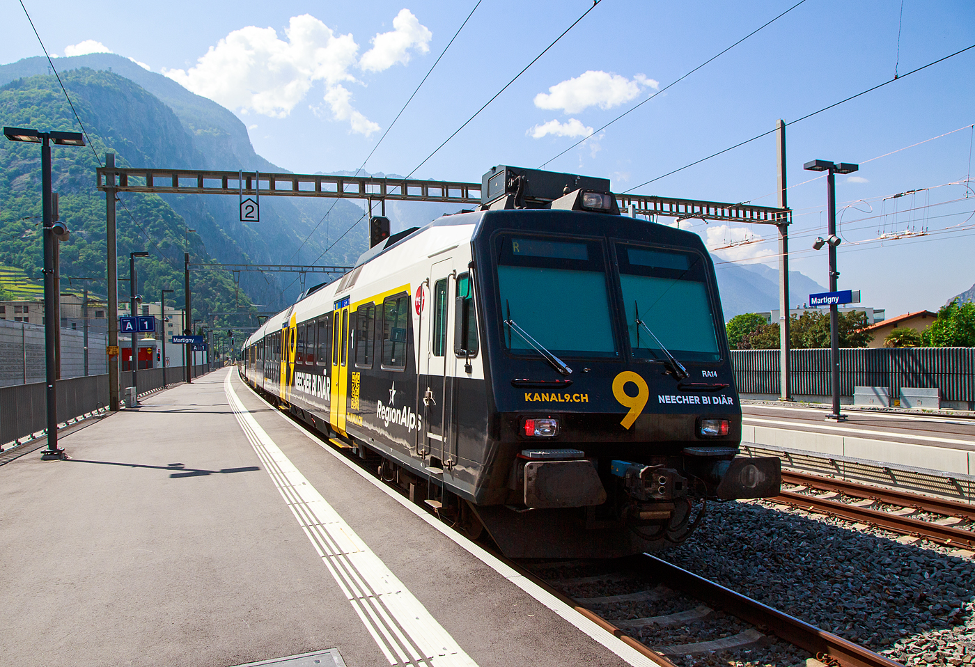 Der dreiteilige NPZ Domino RA14 der RegionAlps, mit der Vollwerbung für den Walliser Fernseh-Sender Kanal 9, verlässt am 26.Mai 2023, als Regionalzug nach Monthey den Bahnhof Martigny.

Der NPZ Domino besteht aus den Triebwagen RBDe 560 DO RA 94 85 7 560 414-5, dem 2.Klasse Wagen B NPZ DO RA 50 85 29-43 414-6 und dem 1./2. Klasse Steuerwagen ABt NPZ DO RA 50 85 39-43 954-9 CH-RA (ex SBB RBDe 560 276, ex SBB B 29-43 181 und ex SBB ABt 39-43 869).

Die Regionalps (RA) ist ein Eisenbahnverkehrsunternehmen im Kanton Wallis, mit Sitz in Martigny hat. Eigentümer derim Jahr 2003 gegründete Gesellschaft  sind die Schweizerischen Bundesbahnen (SBB) mit 70 %, die Transports de Martigny et Régions (TMR) mit 18 % und der Kanton Wallis mit 12 %. Die RA betreibt den Regionalverkehr vor allem im Rhonetal zwischen dem Ostufer des Genfersees und Brig. Inzwischen gehören Regionalps 16 dreiteilige NPZ Domino (Trieb-, Zwischen- und Steuerwagen ex SBB) und 4 RABe 525 NINA (ex TMR).
