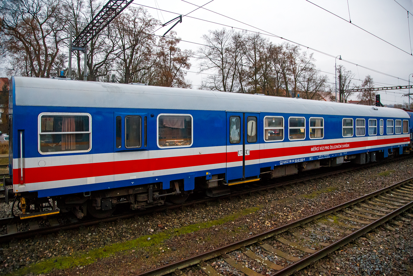 Der Gleismesswagen CZ-SZCZ 99 54 93-62 005-9 der Spr�va �eleznic (S�CZ) steht am 24.11.2022 hinter der Vectron 193 901-6 bei Karl�tejn (deutsch Karlstein, zum Bezirk Beroun). Aufnahme aus dem Zugheraus.

Das staatliche tschechische Eisenbahninfrastrukturunternehmen hie� bis Januar 2020 S�DC - Spr�va �elezničn� dopravn� cesty (deutsch: Verwaltung der Eisenbahn-Verkehrswege) und wurde Spr�va �eleznic (S�CZ) unbenannt.

Der Gleismesswagen dient zur Messung der geometrischen Parameter der Gleise. Er wurde von der niederl�ndischen Firma ER aus einem vom VEB Waggonbau Bautzen 1989/90 gebauten ČD-Schnellzugwagen der Gattung Bymee umgebaut. 

Die geometrischen Parameter des Gleises werden ber�hrungslos alle 0,25 m des abgefahrenen Gleises abgetastet. Die Messgr��energebnisse werden in dynamische und quasistatische Anteile der Gleislage unterteilt. Dynamische Gr��en repr�sentieren L�ngsh�he, �berh�hung, Gleisrichtung, Spurweite, Einbruch mit optionaler Basisl�nge. Quasistatische Gr��en repr�sentieren Kr�mmung, H�he und Spurweite. Das System zur Messung einzelner geometrischer Parameter verwendet Beschleunigungssensoren, Winkelgeschwindigkeiten, induktive Wegsensoren, ein laseroptisches System mit CCD-Kameras (zur Spur- und Richtungsmessung). Die Messwerte werden von einem Computer verarbeitet mit einer �bersicht �ber die unmittelbaren M�ngel der Abschnittsauswertung. Individuelle Dateien erm�glichen die Weiterverarbeitung im Auswertezentrum. Der Messwagen ist au�erdem mit einem Videosystem ausgestattet, das die gemessene Strecke kontinuierlich aufzeichnet.

TECHNISCHE DATEN:
Spurweite: 1.435 mm (Normalspur)
Anzahl der Achsen: 4 
L�nge �ber Puffer:  26.400 mm
Drehzapfenabstand: 19.000 mm
H�he: 4.050 mm
Breite: 3.140 mm
Achsabstand: 8.000 mm
Eigengewicht: 43.245 kg
H�chstgeschwindigkeit: 160 km/h 
Bremse: DK-PR (D)
Kleister befahrbarer Gleisbogen: R = 150 m
