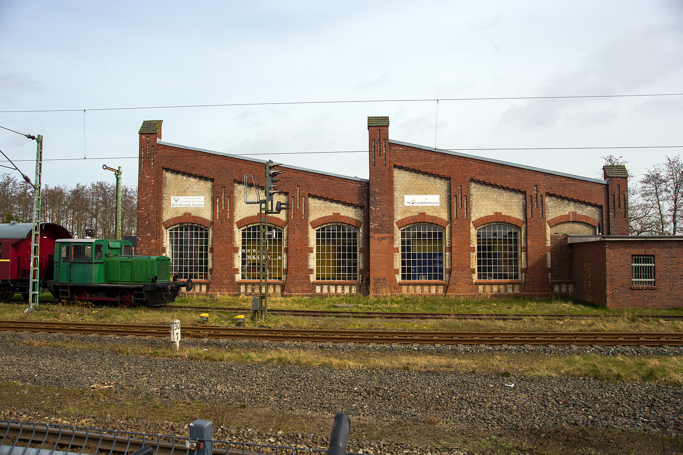 Der Lokschuppen (Seitenansicht/Westseite vom Bf) der Museumseisenbahn K�stenbahn Ostfriesland e. V. am 14 M�rz 2024, gleich neben dem Bahnhof Norden.

Zur Inbetriebnahme der Ostfriesischen K�stenbahn im Jahre 1883 wurde auch die Einrichtung eines Bahnbetriebswerks im Bahnhof Norden notwendig.Neben den erforderlichen Anlagen wurde ein zweist�ndiger Rechteckschuppen errichtet, der sich ungef�hr an der Stelle befand, wo heute das Norder Fahrdienstleiterstellwerk steht. 

Da diese Einrichtung schon bald nicht mehr ausreichte, wurde in den Jahren 1911/12 auf dem gegen�berliegenden Gel�nde ein neuer, vierst�ndiger Rundlokschuppen errichtet. Der Bau des Lokschuppens erfolgte nach einer Musterzeichnung der Preu�ischen Staatsbahn Direktion M�nster, die an den bereits bestehenden Lokschuppen des Bahnbetriebswerks Osnabr�ck angelehnt war. Die Bauausf�hrung erfolgte durch das ortsans�ssige Unternehmen. Die Bauabnahme erfolgte am 8. Januar 1912.

Das Bw verf�gte �ber eine Drehscheibe mit 20 Metern Durchmesser, einen Wasserturm, ein Heizwerk zum Vorheizen der Reisez�ge und des Betriebswerks selbst, eine Bekohlungsanlage mit zwei Drehkr�nen und diverse Nebengeb�ude.

In erster Linie waren Tenderlokomotiven der preu�ischen Baureihen T 5 (BR 71/72) und T 11/12 (BR 74) in Norden beheimatet. Im Jahr 1923 wurden die ersten beiden T 18 (BR 78) zugewiesen. Die Loks bespannten haupts�chlich Z�ge auf der Ostfriesische K�stenbahn (KBS 393) Norden - Jever - Wilhelmshaven.

Im Herbst 1949 verf�gte das Bw �ber 115 Mitarbeiter. Der Unterhaltungsbestand an Lokomotiven sah am 15. M�rz 1950 noch folgende Loktypen vor: zwei Loks der Baureihe 74 und sieben der Baureihe 78. Durch den verst�rkten Einsatz von Schienenbussen zwischen Norden und Wilhelmshaven ab Sommer 1952 verlor das Bw seine Bedeutung. Als selbst�ndige Dienststelle wurde es 1954 aufgel�st und in eine Au�enstelle des Bw Emden umgewandelt.

In der folgenden Zeit wurde die Drehscheibe entfernt und alsbald wurden auch die Bekohlungsanlage und andere zum Bw geh�rende Bauten aufgelassen und abgebrochen. Nach Aufl�sung des Norder Betriebwerkes diente der Lokschuppen den verschiedensten Zwecken, als Fahrzeugunterstell- und Lagerraum f�r die Bahnmeisterei, D�ngemittel-Lager eines Landhandels und Winterquartier f�r Sportboote.

Erst als die MKO im Jahr 1990 das Bauwerk von der Deutschen Bundesbahn anmieten konnte, wurde es wieder f�r seinen urspr�nglichen Zweck zur Unterstellung und Wartung von Eisenbahnlokomotiven genutzt.