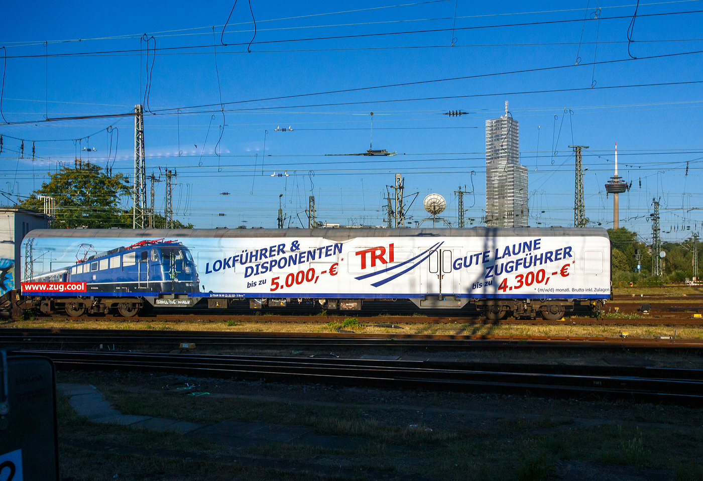 Der Werbewagen D-TRAIN 50 80 22-35 929-5 der Gattung Bnrz 450.3 (Vollwerbung TRI sucht Lokführern/Disponenten und Gute Laune Zugführern) der TRI Train Rental GmbH, abgestellt beim BW Köln Bbf, hier am 01 Juli 2025 aus einem Zug fotografiert. Der Wagen ist ein n-Wagen (ex Silberling) bzw. ein lazarettfähiger 2. Klasse Nahverkehrswagen, er wurde 1970 vom AW Karlsruhe, als DB 50 80 22-35 929-5 Bnrz 724 gebaut. 2001 wurde er im DB Werk Wittenberge in die Gattung Bnrz 450.3 umgebaut. Vermutlich 2017/18 wurde er von der DB Regio an die TRI Train Rental GmbH verkauft. Die Unterbauart 450.3 verfügt im Gegensatz zu den übrigen Varianten weiterhin über eine lazarettfähige Inneneinrichtung.

TECHNISCHE DATEN:
Spurweite: 1.435 mm (Normalspur)
Länge über Puffer: 26.400 mm
Wagenkastenlänge: 26.100 mm
Wagenkastenbreite: 2.825 mm
Höhe über Schienenoberkante: 4.050 mm
Drehzapfenabstand:19.000 mm
Achsstand im Drehgestell: 2.500 mm
Drehgestellbauart: Minden-Deutz 432
Leergewicht: 33 t
Höchstgeschwindigkeit: 140 km/h
Sitzplätze: 96 in der 2. Klasse
Abteile: 3 Großräume (2. Klasse)
Toiletten: 1, geschlossenes System
Dienstraum: ja
Bemerkungen: Design OFV mit Glastrennwänden; lazarettfähig
