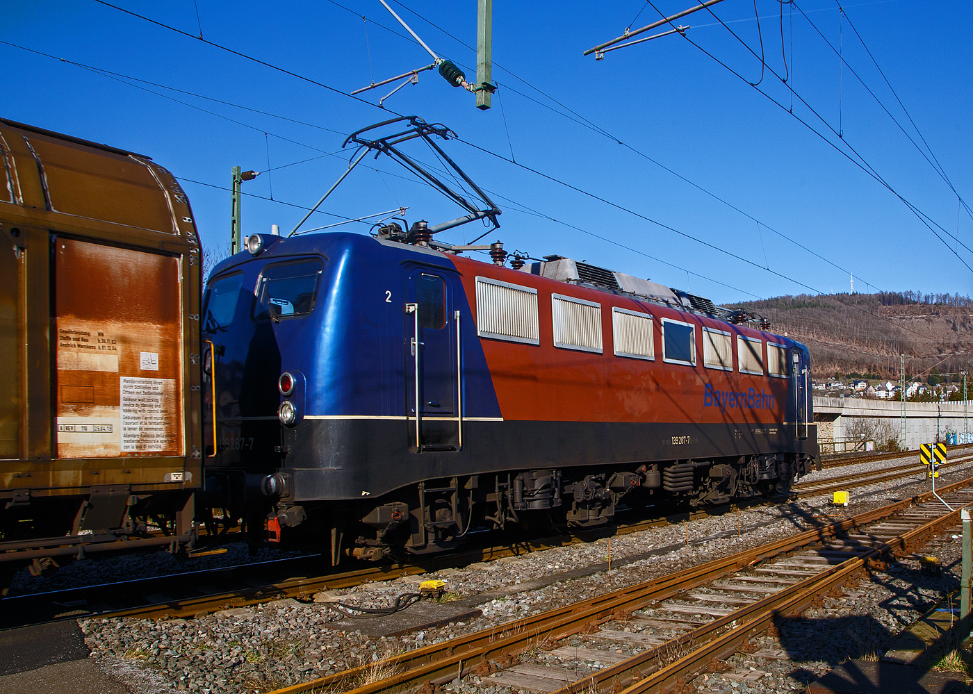 Die 139 287-7 (91 80 6139 287-7 D-BYB) der BayernBahn GmbH fährt am 14.02.2023 mit dem sogenannten  Henkelzug  (Langenfeld/Rhld. nach Gunzenhausen), durch Niederschelderhütte, hier beim Bü 343 (km 112,183) kurz vor Niederschelden bzw. der Landesgrenze zu NRW, in Richtung Siegen.

Nochmal einen lieben Gruß an den netten Lokführer zurück.

Die Lok wurde 1963 von Krauss-Maffei AG in München-Allach unter der Fabriknummer 18956 gebaut, er elektrische Teil ist von den Siemens-Schuckert-Werke (SSW) in Berlin. Als DB E10 287 wurde in Dienst gesetzt, mit der Einführung des EDV-Nummernsystems wurde sie zum 01.01.1968 zur DB 110 287–0. Die Lok ist mit Einholmstromabnehmern ausgerüstet, sie ist die letzte gebaute Kasten 110er. Zur 139er wurde sie erst 1994 nach einem Umbau, der Lokkasten wurde auf Drehgestelle der Baureihe 140 (E40) gesetzt und es folgte die Umzeichnung in DB 139 287-7. Zum Mai 2009 erfolgte die Z-Stellung bei der DB AG. Zum 1.Oktober 2011 wurde die Lok durch die BayernBahn GmbH erworben und erhielt anschließend eine Untersuchung. Die Neulackierung in kobalt-blau/orange erhielt sie dann 2017.
