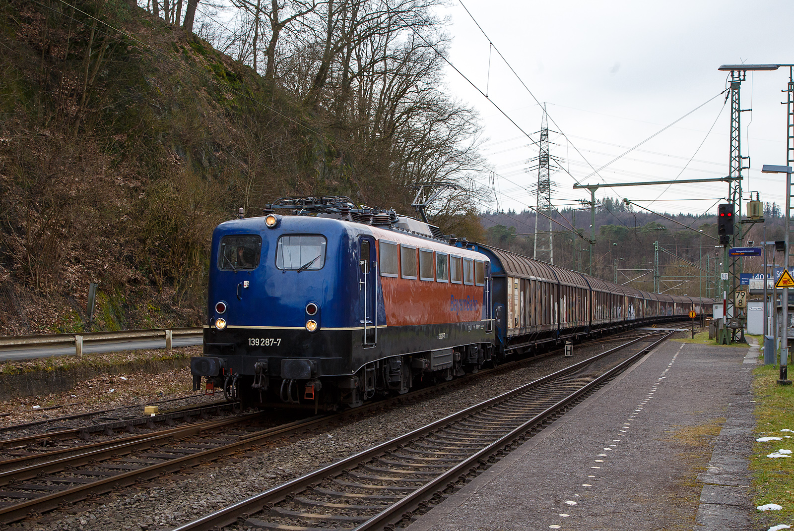 Die 139 287-7 (91 80 6139 287-7 D-BYB) der BayernBahn GmbH fährt am 17.03.2023 mit dem sogenannten  Henkelzug  (Langenfeld/Rhld. nach Gunzenhausen), durch Scheuerfeld (Sieg) in Richtung Siegen. Der Zug bestand aus gedeckten Schiebewand-Wageneinheiten der Gattung Hiirrs.

Nochmal einen lieben Gruß an den netten Lokführer zurück.

Die Lok wurde 1963 von Krauss-Maffei AG in München-Allach unter der Fabriknummer 18956 gebaut, er elektrische Teil ist von den Siemens-Schuckert-Werke (SSW) in Berlin. Als DB E10 287 wurde in Dienst gesetzt, mit der Einführung des EDV-Nummernsystems wurde sie zum 01.01.1968 zur DB 110 287–0. Die Lok ist mit Einholmstromabnehmern ausgerüstet, sie ist die letzte gebaute Kasten 110er. Zur 139er wurde sie erst 1994 nach einem Umbau, der Lokkasten wurde auf Drehgestelle der Baureihe 140 (E40) gesetzt und es folgte die Umzeichnung in DB 139 287-7. Zum Mai 2009 erfolgte die Z-Stellung bei der DB AG. Zum 1.Oktober 2011 wurde die Lok durch die BayernBahn GmbH erworben und erhielt anschließend eine Untersuchung. Die Neulackierung in kobalt-blau/orange erhielt sie dann 2017.