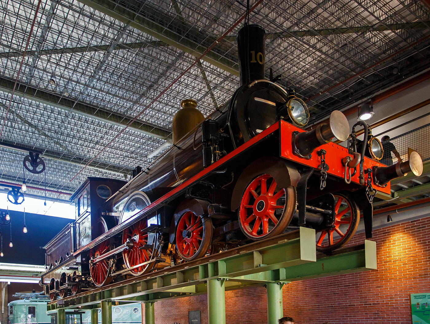 Die 1889 gebaute 2'B Schnellzug-Dampflokomotive NRS 107 „Rhijnboog“ (Rheinbogen), später HSM 353, NS 1604 (NS-Baureihe 1600), am 29 April 2025 hoch aufgeständert im Het Spoorwegmuseum (bis 2005 NSM - Nederlands Spoorwegmuseum / Niederländischen Eisenbahnmuseum) in Utrecht im ehemaligen Bahnhof Maliebaanstation.   

Die Dampflok wurde 1889 von Sharp, Stewart & Co. in Glasgow (GB) unter der Fabriknummer 3563 gebaut und an die NRS - Nederlandsche Rhijnspoorweg-Maatschappij (Niederländische Rheinbahn-Gesellschaft). Im Jahr 1890 wurde die NRS vom niederländischen Staat übernommen, der ihren Betrieb an die Staatsbahnen (SS) übertrug. Das NRS-Rollmaterial wurde zwischen den Staatsbahnen (SS) und der Hollandse IJzeren Spoorweg-Maatschappij (HSM) aufgeteilt, so wurde die NRS 107 zur HSM 353. Ab 1921, nach der Zusammenlegung des Rollmaterials der SS und der NRS, wurde die Lok zur NS 1604.

Die Baureihe NS 1600 war eine Reihe von 59 Schnellzugdampflokomotiven der NS - Nederlandse Spoorwegen (Niederländischen Eisenbahnen) und ihrer Vorgängergesellschaften, der NRS - Nederlandsche Rhijnspoorweg-Maatschappij (Niederländische Rheinbahn-Gesellschaft), der SS - Maatschappij tot Exploitatie van Staatsspoorwegen (Gesellschaft zur Nutzung der Staatseisenbahnen) und der HSM - Hollandsche IJzeren Spoorweg-Maatschappij (Niederländischen Eisenbahn-Gesellschaft). Die Loks wurden von Sharp, Stewart & Co. in Glasgow (GB) gebaut.

Die Loks vom Typ „Rhijnboog“ stellt einen großen Sprung nach vorn im Lokbau dar. Es war die erste Lokomotive, die vorne ein führendes Lauflachsen-Drehgestell hatte, das die Fahrleistung verbesserte. Dies ermöglichte auch die Länge und damit die Kapazität des Kessels zu erhöhen. Sie gehörten damals zu den größten Lokomotiven Europas.

Der Spitzname  Rhijnboog  kombiniert den Namen der niederländischen Eisenbahngesellschaft, die die Lokomotive (Nederlandsche Rhijnspoorweg-Maatschappij) mit dem Wort  Bogie  bestellt hat. Nach der Auflösung der NRS wurde die Lokserie schließlich von der HSM übernommen. Sie waren mit diesen Loks so zufrieden, dass sie 50 weitere nahezu identische Lokomotiven bestellte. Die Lokomotive NRS 107 hat einen besonderen Platz im Museum, sie überragt die Besucher von seinem erhabenen  Thron . 

NRS 101 bis 109:
Im Jahr 1889 bestellte die Nederlandsche Rhijnspoorweg-Maatschappij (NRS) neun Lokomotiven mit den Nummern 101–109 bei der Fabrik von Sharp Stewart and Company in Glasgow. Die Achsfolge der Lokomotiven war 2'B. Zum Zeitpunkt der Bestellung waren dies die ersten niederländischen Lokomotiven mit einem Laufdrehgestell vor den Antriebsrädern (Triebachsen). Sie waren die letzten von der NRS bestellten Lokomotiven. Ein Jahr später, 1890, wurde die NRS aufgelöst und die Ausrüstung zwischen der Niederländischen Eisenbahn-Gesellschaft (HSM) und der Staatlichen Eisenbahn-Ausbeutungsgesellschaft (SS) aufgeteilt. Die Lokomotiven 101, 102, 103, 108 und 109 gingen an die SS und erhielten die SS-Nummern 1101–1103 und 1108–1109. Die 104-107 gingen an das HSM und bekamen die HSM-Nummern 350-353. Da die Lokomotiven für die kleinen Drehscheiben der SS zu lang waren, übergab die SS die fünf Lokomotiven ein Jahr später, 1891, an die HSM, wo sie die Nummern 354–358 erhielten. Aufgrund des Drehgestells für die Antriebsräder, auch Bogie genannt, und der Herkunft des Rhijnspoor erhielten diese Maschinen bei der HSM den Spitznamen „Rhijnboog“.

HSM 359 bis 408:
Die HSM war mit den von ex NRS Lokomotiven so zufrieden, dass sie 40 weitere bei Sharp Stewart and Company bestellte. Diese wurden zwischen 1891 und 1892 unter den Nummern 359–398 in Dienst gestellt. Im Jahr 1900 wurde eine weitere Bestellung über 5 Einheiten mit den Nummern 399-403 bei Sharp Stewart and Company aufgegeben. Schließlich wurde im Jahr 1903 die Klasse 404–408 in Betrieb genommen, die von der North British Locomotive Company (Atlas Works) gebaut wurden, da Sharp Stewart & Co. Company inzwischen zu dieser Gesellschaft fusioniert war.

Bedingt durch den Ersten Weltkrieg wurde 1917 eine Zweckgemeinschaft der unabhängigen Eisenbahngesellschaften HSM (Hollandsche IJzeren Spoorweg-Maatschappij) und SS (Maatschappij tot Exploitatie van Staatsspoorwegen) gegründet. Die beiden Gesellschaften errichteten einen gemeinsamen Bahnbetrieb, blieben aber rechtlich unabhängig. Dies änderte sich 1938, als HIJSM und SS vollständig fusionierten und in der AG NS (Nederlandse Spoorwegen, früher Nederlandsche Spoorwegen geschrieben) aufgingen. Das Rollmaterial wurde bereits 1921 zusammengelegt, so wurden die Lok zur NS 1604 nummeriert.

Die NS 1614, 1633, 1636, 1637 und 1646 wurden 1921 mit einem Überhitzer ausgestattet. Es war beabsichtigt, die gesamte Serie damit auszurüsten, aber aufgrund enttäuschender Ergebnisse blieb es bei diesen fünf Lokomotiven. Sie wurden in den Jahren 1936–1949 außer Dienst gestellt. Die Lokomotive NS 1622 wurde als letzte dieser Baureihe im Jahr 1949 ausgemustert. Die NS 1604, ex HSM 353, ex NRS 107 „Rhijnboog“ blieb bis 1940 im Einsatz blieb. Die 1604 wurde zur Erhaltung für das Niederländische Eisenbahnmuseum in Utrecht ausgewählt und im Lokschuppen in Maastricht eingelagert, wo die Lokomotive am 8. September 1944 durch Kriegseinwirkung schwer beschädigt wurde. Nach dem Zweiten Weltkrieg wurde die Lokomotive von der NS-Hauptwerkstatt in Tilburg vollständig restauriert und in die braune Version der NRS mit der ursprünglichen Nummer 107 zurückversetzt. Seit März 1955 ist die NRS 107 im Eisenbahnmuseum ausgestellt. Nach der Sanierung des Eisenbahnmuseums in den Jahren 2003 bis 2005 wurde die Lokomotive aufgeständert.	

TECHNISCHE DATEN:
NRS-Serie: Rhijnboog, ab 1921 NS 1600
Hersteller: Sharp, Stewart & Co. in Glasgow (GB), später Atlas Works
Gebaute Anzahl: 59
Spurweite: 1.435 mm (Normalspur)
Achsfolge: 	2'B (engl. 4-4-0), gekuppelt mit dreiachsigem Tender
Länge über Puffer: 16.585 mm 
Treibraddurchmesser: 2.016 mm (neu) 
Laufraddurchmesser (Lok und Tender): 1.105 mm (neu) 
Breite: 2.222 mm
Höhe: 4.267 mm
Höchstgeschwindigkeit: 90 km/h
Rostfläche: 2,15 m²
Gesamtheizfläche: 111,6  m²
Kesselüberdruck: 10,3 kg/cm²
Anzahl der Zylinder: 2 (Ø 457 mm x 660 mm Hub)
Steuerung: Stephenson
Eigengewicht: 81,3 t (Lok 48,8 t und Tender 32,5 t) 
Anhängelast: 4.950 kg
Wasservorrat: 10,2 m³ 
Kohlevorrat: 2,8 t
