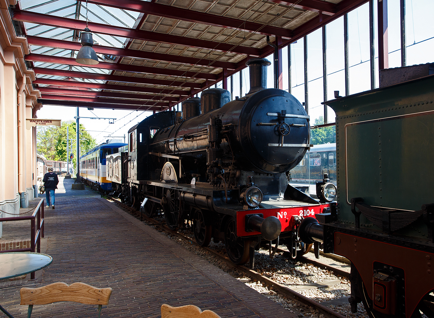 Die 1914 gebaute 2'B Schnellzug-Dampflokomotive NS N° 2104 „Blikken Tinus“ (blecherner Martin) der NS-Baureihe 2100, ex HSM 504, am 29 April 2025 im Het Spoorwegmuseum (bis 2005 NSM - Nederlands Spoorwegmuseum / Niederländischen Eisenbahnmuseum) in Utrecht im Bahnhof Maliebaanstation.   

Die Dampflok wurde 1914 von der BMAG - Berliner Maschinenbau-Actien-Gesellschaft vormals L. Schwartzkopff in Berlin unter der Fabriknummer 5304 gebaut und als HSM 504 an die Hollandse IJzeren Spoorweg-Maatschappij (Holländische Eisenbahn-Gesellschaft) geliefert. 

Die Lokomotiven der Baureihe NS 2100 waren Schnellzugdampflokomotiven der Nederlandse Spoorwegen (NS), die von der Hollandsche IJzeren Spoorweg-Maatschappij (HSM) übernommen wurden.

Die Lokomotiven hatten die Achsfolge 2’B. Die Serie wurde vom HSM-Ingenieur Willem Hupkes für die Beförderung der immer schwereren Personen- und Schnellzüge konzipiert. Die ersten 5 Loks (HSM 501 – 505) wurden 1914 von der BMAG in Deutschland gebaut, 1916 bis 1919 folgten 15 Stück (516 – 530) von Werkspoor in Amsterdam (NL), 1920 folgten 10 Stück (506-515) von der BMAG aus Deutschland, sowie 5 Stück (531 – 535) von Werkspoor aus Amsterdam.

Da das Konkurrenzunternehmen der HSM, die SS - Maatschappij tot Exploitatie van Staatsspoorwegen (Gesellschaft zur Nutzung der Staatseisenbahnen) zu stärkeren 2’C-Maschinen der Serie SS 700 übergegangen war, konnte die HSM nicht zurückbleiben. Obwohl ein HSM-Entwurf für eine 2’C-Maschine bekannt ist, wurde letztlich eine 2’B-Lokomotive ausgewählt. Dies lag jedoch nicht an der begrenzten Länge der Drehscheiben, wie oft fälschlicherweise angenommen wird. Die Maschinen hatten jedoch die maximal zulässige Achslast von 17 Tonnen und die größten Antriebsräder (Ø 2.100 mm) in den Niederlanden, weshalb sie ideal für Schnellzüge waren. Wegen des rasselnden Geräuschs der Kuppelstangen wurden sie vom Personal „Blikken Tinus“ – deutsch blecherner Martin – genannt. Die Lokomotiven wurden von der Berliner Maschinenbau-AG vormals L. Schwartzkopff (BMAG) in Berlin und Werkspoor in Amsterdam gebaut.

Bedingt durch den Ersten Weltkrieg wurde 1917 eine Zweckgemeinschaft der unabhängigen Eisenbahngesellschaften HSM (Hollandsche IJzeren Spoorweg-Maatschappij) und SS (Maatschappij tot Exploitatie van Staatsspoorwegen) gegründet. Die beiden Gesellschaften errichteten einen gemeinsamen Bahnbetrieb, blieben aber rechtlich unabhängig. Dies änderte sich 1938, als HIJSM und SS vollständig fusionierten und in der AG NS (Nederlandse Spoorwegen, früher Nederlandsche Spoorwegen geschrieben) aufgingen. Das Rollmaterial wurde bereits 1921 zusammengelegt, so wurden die Lok zur NS 2104 nummeriert. Die Lokomotive 2104, ehemals HSM 504, wurde als letzte dieser Baureihe 1954 ausgemustert und blieb im Nederlands Spoorwegmuseum erhalten.

TECHNISCHE DATEN:
Hersteller: BMAG in Berlin und Werkspoor in Amsterdam
Baujahre: 1914 bis 1920
Gebaute Anzahl: 35
Spurweite: 1.435 mm (Normalspur)
Achsfolge: 	2'B (engl. 4-4-0), gekuppelt mit dreiachsigem Tender
Länge über Puffer: 17.845 mm 
Treibraddurchmesser: 2.100 mm (neu) 
Laufraddurchmesser (Lok): 990 mm (neu) 
Laufraddurchmesser (Tender): 1.105 mm (neu)
Höhe: 4.370 mm
Höchstgeschwindigkeit: 110 km/h
Dienstgewicht: 105 t (61 t Lok und 44 t Tender)
Anfahrzugkraft: 7.660 kg
Rostfläche: 2,4 m²
Gesamtheizfläche: 154 m² (Strahlungs- 12 m², Rohr- 107 m² und Überhitzer- 35 m²
Kesselüberdruck: 12,4 kg/cm²
Anzahl der Zylinder: 2 (Ø 530 mm x 660 mm Hub)
Steuerung: Walschaerts
Wasservorrat: 19 m³ 
Kohlevorrat: 6,0 t

