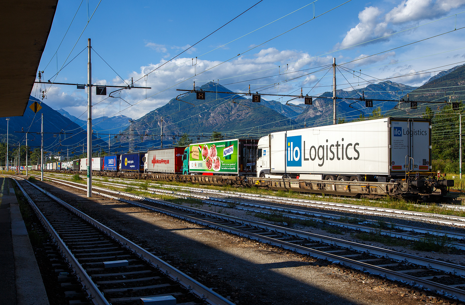 Die an die SBB Cargo International AG vermietete Vectron 193 478-5  Gottardo  und die SBB Cargo Re 484 017-9 / E 484.017 SR haben am 02 August 2019 mit einem RAlpin-RoLa-Zug aus Novara den Rbf Domodossola erreicht. Die Loks kann man links hinten noch gerade erkennen. Obwohl diese Loks den Zug auch weiter durch den Simplon und die Schweiz ziehen k�nnten, die Zulassungen haben sie, �bernehmen gleich zwei BLS Re 485 (TRAXX F140 AC1) den Zug. Aber lange kann man dieses Schauspiel in Domodossola nicht mehr sehen, Ende 2025 wird die Rollende Autobahn eingestellt. 

Diese Rola-Z�ge sind (bald waren) f�r den Transport von Last- und Sattelz�gen im begleiteten Verkehr, einer Form des kombinierten Ladungsverkehrs, vorgesehen und kommt in alpenquerenden Shuttlerelationen zum Einsatz. Begleiteten Verkehr hei�t, dass der ganze Sattelzug oder Anh�ngerz�ge mit max. 44 t und einer max. L�nge von 18,60 Meter (in Ausnahmef�llen 19,0 m), mit der Zugmaschine, auf den Zug verladen werden. Dabei fahren die Lastkraftwagen in speziellen Terminals direkt von der Stra�e �ber eine Rampe auf den Wagen, um vom Ausgangsort zum Ziel transportiert zu werden, auch der LKW-Fahrer f�hrt mit. Die Fahrer ziehen sich in einen speziellen Begleitwagen mit Liegeabteilen (Bmc) zur�ck. 

Am Ziel-Terminal angekommen �bernimmt jeder Fahrer wieder seinen LKW und f�hrt diesen wieder �ber eine Rampe vom Zug auf die Stra�e und liefert die Ware sicher beim Kunden ab. Ein gro�er Vorteil ist auch bei den Fahrern die Einhaltung der „Lenk und Ruhezeiten“. Bei kurzen Shuttlezugverbindungen als Lenkpause gewertet und auf den Mittel- und Langstrecken einen Teil oder sogar als gesamte Ruhezeit gewertet. Dabei trotzdem n�her und ausgeschlafen an Ihr Ziel. Zudem gibt es f�r die Fahrer ein On-Board Service mit Speisen und Getr�nken. 

Weitere Vorteile sind:
•	die Reduzierung des Treibstoffverbrauches und Betriebskilometer
•	die Einsparung der diversen Mauten und Sondermauten
•	die Steigerung des Umsatzes und der Produktivit�t
•	den Einsatz des Lkw trotz Wochenend-, Feiertags-, Nacht- und sonstiger Fahrverbote
•	ersparen von Stauwartezeiten, �berholverbote und andere Behinderungen
•	die Verbesserung der �KO-Bilanz des Unternehmens (CO2-Ersparnis)
•	hohe Sicherheitsstandards der Z�ge
•	Einhaltung von Lenk und Ruhezeiten der Fahrer (allein) auch bei gro�en Entfernungen

Diese Form des kombinierten Verkehrs von Schiene und Stra�e – Rollende Landstra�e (ROLA) – gewann, nicht zuletzt aufgrund von gesteigertem Verkehrsaufkommen und Umweltbewusstsein, immer mehr an Bedeutung. In �sterreich bleibt es wohl auch so, aber hier Italien – Schweiz – Deutschland ist leider Ende 2025 Schluss damit, sehr schade. Obwohl die RAlpin noch Ende 2023 vermeldet hat: knapp 12% mehr Lastwagen per Bahn
umweltfreundlich durch die Schweizer Alpen transportiert als im Vorjahr. 

Die Medienmitteilung RAlpin AG vom 05. Mai 2025 (komplett zitiert):
Die ‚Rollende Autobahn’ wird Ende 2025 eingestellt.

Wegen unerwartet vielen Einschr�nkungen auf dem Schienennetz kann das
Unternehmen RAlpin die ‚Rollende Autobahn‘ (Rola), d. h. den Bahnverlad von ganzen Lastwagen f�r die Fahrt durch die Alpen, auch mit den Finanzhilfen des Bundes nicht mehr wirtschaftlich betreiben, obwohl die Nachfrage von Seiten der Kunden weiterhin gut ist. In Absprache mit dem Bund wird die Rola deshalb fr�her eingestellt als geplant, n�mlich per Ende 2025 statt wie bisher vorgesehen Ende 2028.

Die RAlpin AG steht vor erheblichen wirtschaftlichen Herausforderungen. Trotz der laufenden finanziellen Abgeltungen des Bundes, einer vorhandenen Nachfrage und guter Auslastung von 80 Prozent ist der Betrieb der ‚Rollenden Autobahn’ (Rola) nicht mehr wirtschaftlich m�glich. Bereits 2024 fielen rund 10 Prozent der Z�ge aus. Der Grund waren geplante sowie kurzfristig angeordnete Baustellen und weitere unvorhersehbare Ereignisse. Dies f�hrte 2024 zu einem negativen Ergebnis von rund -2.2 Millionen Franken. Im 1. Quartal 2025 konnten wegen Bauarbeiten im Vergleich zum Vorjahr rund 20 Prozent weniger Z�ge gefahren werden; statt 1018 Z�ge im 1. Quartal sind dieses Jahr in der gleichen Zeitperiode nur deren 794 gefahren.

Das eidgen�ssische Parlament hatte vor zwei Jahren beschlossen, die finanzielle F�rderung der Rola durch den Bund ein letztes Mal zu verl�ngern und den Betrieb per Ende 2028 einzustellen. Die RAlpin kommt nun zum Schluss, dass die Rola unter den ge�nderten Voraussetzungen nicht mehr wirtschaftlich betrieben werden kann, zumal eine Normalisierung der Situation nicht in Sicht ist. In Absprache mit dem Bund hat sie deshalb beschlossen, den Betrieb bereits auf den im Dezember 2025 einzustellen. Die drei Aktion�re der RAlpin (BLS, Hupac und SBB) sind bereit, bis dahin die Finanzierung der Rola sicherzustellen, um eine geordnete Betriebseinstellung zu erm�glichen und die gesch�ftlichen Verpflichtungen zu erf�llen. Der Bund erh�ht seinerseits bis zur Einstellung der Rola die durchschnittliche Abgeltung je verlagerten Lastwagen.

Bei RAlpin sind 16 Personen angestellt, die RAlpin sucht nach L�sungen f�r die betroffenen Mitarbeitenden. Da bereits seit l�ngerem klar ist, dass der Betrieb bis maximal Ende 2028 weitergef�hrt wird, ist RAlpin im steten Austausch �ber den individuellen k�nftigen Berufsweg von allen Mitarbeitenden.

Die Herausforderungen im alpenquerenden kombinierten Verkehr sind gro�:
Die RAlpin hat in den letzten Jahren die notwendigen Voraussetzungen geschaffen, trotz Kostensteigerungen f�r die Nutzung der Bahninfrastruktur, Energie, Terminals und Traktion mit einem konstanten Zahlungsrahmen j�hrlich bis zu 80.000 Lastwagen im begleiteten kombinierten Verkehr durch die Alpen auf die Schiene zu verlagern. Dies entspricht 7 Prozent des kombinierten Verkehrs durch die Schweizer Alpen. Die RAlpin wird gemeinsam mit den Kunden M�glichkeiten suchen, um ihre Mengen weiterhin auf der Schiene zu transportieren. Es ist aber davon auszugehen, dass ein Teil, der auf der Rola transportierten G�ter zuerst auf die Stra�e zur�ck verlagert wird, bis die Umr�stung auf kranbare Sattelauflieger erfolgt ist. Wobei es gibt Systeme bei denen die Sattelauflieger nicht kranbar sein m�ssen.

Die Situation der Rola ist symptomatisch f�r den ganzen alpenquerenden kombinierten Verkehr auf der Schiene. Grund f�r die zunehmenden Zugausf�lle auf der Rola ist insbesondere die anhaltend hohe St�rungsanf�lligkeit der Schieneninfrastruktur in Deutschland. Die RAlpin erwartet, dass sich die Bedingungen f�r die Akteure im kombinierten Verkehr in den kommenden Jahren verbessern werden und so neue tragf�hige L�sungen m�glich sind, um das Verlagerungsziel von der Stra�e auf die Schiene zu erreichen. Dazu wird es weiterhin flankierende Ma�nahmen durch die Politik brauchen. Es muss das Ziel sein, die Anstrengungen der letzten 25 Jahre f�r eine erfolgreiche Verlagerung des alpenquerenden Verkehrs durch die Schweiz auf die Schiene langfristig zu sichern.

So soll langfristig der unbegleitete kombinierter Verkehr (ohne LKW-Zugmaschine) die RoLa abl�sen: 
Die ‚Rollende Landstrasse’ (Rola) war seit den sp�ten 1960er-Jahren das erste Angebot, um den G�terschwerverkehr durch die Alpen von der Stra�e auf die Schiene zu verlagern. Dabei werden ganze Lastwagen oder Sattelschlepper auf die Schiene verladen und die LKW-Chauffeure reisen in einem Begleitwagen im Zug mit. Daher ist die Rola auch als „begleiteter kombinierter Verkehr“ bekannt.

Im Zuge des Ausbaus der Alpentransitstrecken wurde die Rola 2001 mit der neu gegr�ndeten RAlpin AG, einem Gemeinschaftsunternehmen von SBB, BLS und Hupac, modernisiert. Die Betreiberin transportiert Lastwagen auf der Strecke zwischen Freiburg im Breisgau (D) und Novara in Italien. Das Angebot war von Beginn weg als �bergangsl�sung konzipiert, bis die Fertigstellung der NEAT einen effizienten Bahntransport von Sattelaufliegern (Sattelschlepper ohne Zugfahrzeug und Chauffeur) und Containern �ber lange Distanzen erlaubt. Das eidgen�ssische Parlament hatte vor zwei Jahren beschlossen, die finanzielle F�rderung der RoLa durch den Bund ein letztes Mal zu verl�ngern und den Betrieb per Ende 2028 einzustellen.

Parallel dazu entwickelte sich der „unbegleitete Kombinierte Verkehr“ dank fortschrittlichen Umschlagtechniken wie kranbaren Sattelauflegern und Containern weiter. Wobei bei es gibt heute schon Systeme bei denen nicht kranmanipulierbare Sattelauflegern auch im KLV gefahren werden k�nnen. Erw�hnenswert sind da u.a. das System „NiKRASA“ der TX Logistik AG, das weltweit patentierte System Trailer-Wagen der Helrom GmbH, sowie das horizontale Verladesystem der CargoBeamer AG. Mit der Fertigstellung der NEAT geh�rt die Zukunft auf dem Nord-S�d-Korridor dieser Sparte des kombinierten Verkehrs.

RAlpin AG auf einen Blick
Die RAlpin AG mit Sitz in Olten betreibt die Rollende Autobahn (Rola) zwischen Freiburg i. Br. und Novara. Ihr Angebot umfasst j�hrlich 100.000 Verladegelegenheiten f�r Lastwagen von der Stra�e auf die Schiene. Im 2024 wurde das Angebot von rund 72.000 Lastwagen genutzt. Die Lastwagenfahrer/-innen verladen ihre Fahrzeuge in den Terminals auf Niederflurtragwagen und reisen sicher in einem Begleitwagen mit. Mit diesem Angebot leistet RAlpin einen Beitrag zur Verkehrsverlagerung von der Stra�e auf die Schiene und zum Schutz der sensiblen Alpenregion. (Zusatz von mir: Nur leider spielt Deutschland nicht so mit).

Die Aktion�re der RAlpin sind BLS AG, Hupac SA und SBB AG.

Quellen: RAlpin, Railcargo
Stand: September 2025

