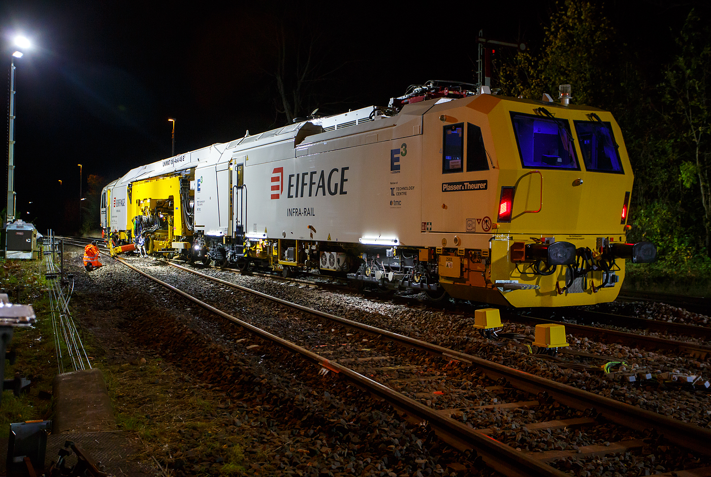 Die Baustelle Bahnhof Herdorf am Abend des 26 Oktober 2025, nun ab 20:40 Uhr wird die neue Weiche 3, beim Bahnhof/Stellwerk Herdorf Fahrdienstleiter (Hf), gestopft. Hier kam die recht neue Plasser & Theurer UNIMAT 09-4x4/4S E� (A-PLA 99 81 9124 004-8), eine Universal-Weichenstopfmaschine mit Hybrid-Antriebstechnik, der Firma Eiffage Infra-Rail GmbH (Herne) zum Einsatz. Ob die Maschine nur von Eiffage Infra-Rail gemietet oder nun gekaut wurde ist mir nicht klar, aber eingestellt ist sie in �sterreich �ber Franz Plasser Vermietung von Bahnbaumaschinen GmbH. 

Die Maschine wurde 2017 von Plasser & Theurer in Linz unter der Fabriknummer 6557/58 gebaut. Diese Maschine mit der neuen E�-Antriebstechnologie von Plasser & Theurer wurde erstmals 2017 auf der iaf 2017 in M�nster pr�sentiert, wo ich sie am 01.06.2017 auch ablichten und sogar in ihr Platz nehmen konnte. Siehe u.a.:
http://hellertal.startbilder.de/bild/oesterreich~bahndienstfahrzeuge~stopfmaschinen/572409/neues-maschinendesign-und-neue-e179-antriebstechnologie-von.html
und
http://hellertal.startbilder.de/bild/oesterreich~bahndienstfahrzeuge~stopfmaschinen/572406/der-blick-des-maschinenfuehrers-auf-das.html

Leider ist die Helltalbahn nicht elektrifiziert, somit konnte die Maschine den Bahnhof Herdorf nur mit Dieselantrieb (diesel-elektrisch) anfahren und nur im Dieselbetrieb arbeiten. Wenn aber eine Oberleitung vorhanden ist, dann kann die Maschine auch rein elektrisch arbeiten. 

Die Fahrantriebe sind elektrisch im Dieselbetrieb diesel-elektrisch. Im Arbeitsmodus werden alle Drehbewegungen elektrisch, Linearbewegungen hydraulisch. Zudem wir im Dieselbetrieb �bersch�ssige elektrische Energie ins Netz zur�ck gespeist. 

Der diesel-elektrisch angetriebener Fahrmodus und Arbeitsmodus:
Ein Dieselmotor mit einer Leistung von 600 kW liefert die Energie f�r die elektrische Versorgung der Antriebskomponenten �ber den Trafo und Stromrichter. Er wirkt dabei als Generator. Im Arbeitsbetrieb funktionieren die Aggregate gleicherma�en wie im elektrischen Modus. Wichtigster Vorteil ist die Reduktion von der CO2-Emission und von L�rm (10 dBA L�rmreduzierung). Weniger Triebstoffverbrauch, h�here Energieeffizienz im Diesel-elektrischen Betrieb. Ich konnte pers�nlich feststellen, dass die Maschine (auch beim Stopfen) erheblich leiser ist, als andere klassische Maschinen. 

TECHNISCHE DATEN:
Fahrzeugbegrenzungsprofil: G1
Spurweite: 1.435 mm (Normalspur)
Anzahl der Achsen: 8 (in 4 Drehgesten)
L�nge �ber Puffer:  34.140 mm
Drehzapfenabst�nde: 11.250 mm / 4.550 mm / 10.300 mm
Achsabstand im Drehgestell: 1.800 mm
Raddurchmesser (neu): 920 mm
Eigengewicht: 142.000 kg
Kleister befahrbarer Gleisbogen: R = 150 m 
Zugelassen f�r Streckenklasse: C2 und h�her
H�chstgeschwindigkeit: 100 km (eigen und geschleppt)
Kraftstofftank: max. 2.580 l (Diesel)
Wirkungsgrad: bis zu 94 %
Bremse:2 x KE-GP mZ (K)
Bremssohle: J 816 M