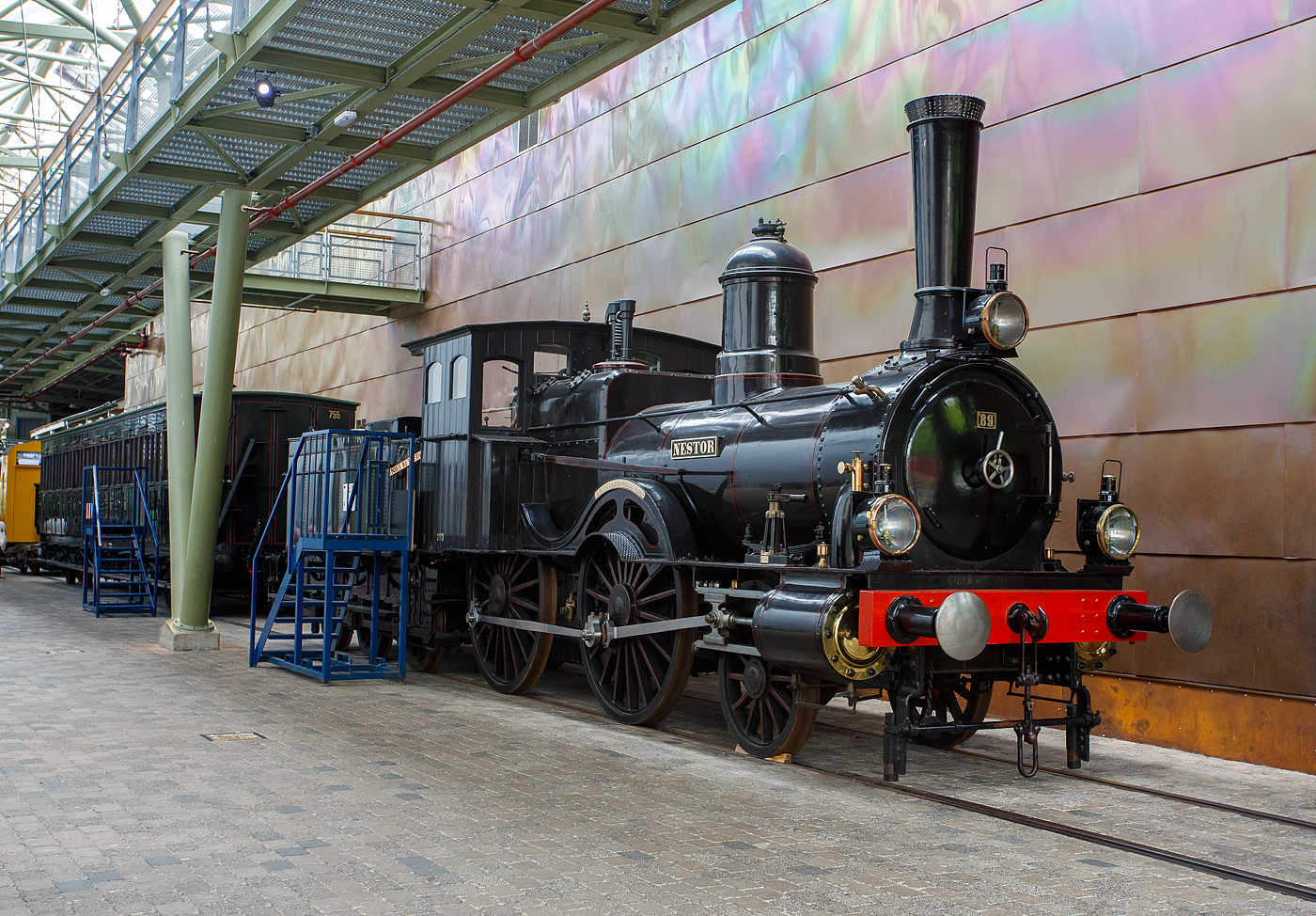 Die Dampflokomotive HSM 89  Nestor , sp�ter NS № 1010 (NS-Baureihe 1000) am 24 Juni 2025 im Het Spoorwegmuseum (bis 2005 NSM - Nederlands Spoorwegmuseum / Niederl�ndischen Eisenbahnmuseum) in Utrecht im ehemaligen Bahnhof Maliebaan.   

Die Dampflok wurde 1879 von Borsig in Berlijn-Tegel unter der Fabriknummer 3730 gebaut und an die damalige HSM - Hollandsche IJzeren Spoorweg-Maatschappij (Holl�ndische Eisenbahn-Gesellschaft) geliefert, 1880 erfolgte die Inbetriebnahme.

HSM 89  Nestor  ist eine niederl�ndische 1B (2-4-0) Tenderlokomotive, die vom Niederl�ndischen Nationalbahnmuseum erhalten wurde. 29 seiner Art wurden f�r den NS-Vorg�nger Hollandsche IJzeren Spoorwegmaatschappij (HSM) gebaut.

In den fr�hen Tagen war die HSM die einzige Eisenbahngesellschaft in den Niederlanden, die ihren Lokomotiven immer einen Namen sowie eine Nummer gab. Der Nestor zum Beispiel wurde auch die Zahl 89 zugewiesen. Trotz der Tatsache, dass es immer als  Der Nestor  bezeichnet wurde, zeigt das Namensschild nur  Nestor . Diese Serie von Lokomotiven wurde  Tenners  genannt, weil sie mit Dampfdruck in H�he von 10 Atmosph�ren umgehen konnten. Ein bemerkenswertes Merkmal dieser Lokomotivserie ist, dass der F�hrerstand der aus Holz und nicht aus Metall besteht. Ihre Lokf�hrer und Heizer wurden daher manchmal  Woonwagenbewoners  genannt, was ungef�hr  Caravan-Bewohner  bedeutet. Metallstangen, die als Schienenw�chter bekannt sind, wurden an der Vorderseite der Rahmenplatten befestigt. Der Lokf�hrer konnte Besen zu diesen Wachen montieren, um die Schienen unter verschneiten Bedingungen zu r�umen. Wie alle Lokomotiven hat der Nestor auch Fettt�pfe montiert. Diese T�pfe w�ren mit Fett und Garn gef�llt oder gef�hlt, um die Achsen zu schmieren.

HSM
Mit dem Ausbau einer Eisenbahnlinie und der Er�ffnung einer weiteren 1879 wurde die Notwendigkeit, ihre Sammlung von Lokomotiven zu erweitern, deutlich und bestellte 29 dieser Maschinen in Borsig. Sie waren 74-82, 89-98 und 116-125 nummeriert, weil die HSM Lokomotiven anhand des Empfangsauftrags nummerierte. Die Serie hie� EOS - JORDAENS, die Namen von 74 und 125. Trotz der Namensschilder, die nur einen Namen haben, wurden sie in der Sprache immer als Der Name bezeichnet (z. B.  Der Nestor  statt  Nestor ).

Bei ihrer Ankunft erhielten verschiedene �ltere Lokomotiven weniger wichtige Arbeiten, die noch �ltere Lokomotiven aus dem Dienst zogen. Mit der Ankunft neuer Sharp Stewart-Lokomotiven im Jahr 1890 wurden die EOS - JORDAENS auf sekund�re, weniger wichtige Aufgaben reduziert, wie sie es zuvor bei �lteren Lokomotiven getan hatten. Sie arbeiteten in den Depots Almelo, Amsterdamer Hauptbahnhof, Apeldoorn, Haarlem und Zutphen und den meisten gro�en Stationen entlang einer Linie nach Osten nach Westen mitten im Land. Sp�ter verteilten sie sich noch mehr auf andere HSM-Depots, die immer mehr auf Rangierarbeiten reduziert wurden.

NS
Bedingt durch den Ersten Weltkrieg wurde 1917 eine Zweckgemeinschaft der unabh�ngigen Eisenbahngesellschaften HSM beziehungsweise HIJSM (Hollandsche IJzeren Spoorweg-Maatschappij) und SS (Maatschappij tot Exploitatie van Staatsspoorwegen) gegr�ndet. Die beiden Gesellschaften errichteten einen gemeinsamen Bahnbetrieb, blieben aber rechtlich unabh�ngig. Dies �nderte sich 1938, als HIJSM und SS vollst�ndig fusionierten und in der AG NS (Nederlandse Spoorwegen, fr�her Nederlandsche Spoorwegen geschrieben) aufgingen. Das Rollmaterial wurde bereits 1921 zusammengelegt, so wurden die EOS - JORDAENS zu NS 1001 - 1029 nummeriert. Die NS vergr��erte auch die Fenster in der Kabine.

Bis 1923 befanden sich acht von ihnen am Hauptbahnhof Utrecht, der vor der Fusion ausschlie�lich Staatsspoorwegen-Gebiet war. Sie wurden ausschlie�lich zum Rangieren verwendet. In Amersfoort waren f�nf weitere mit der gleichen Arbeit und in Winterswijk, vier, obwohl diese vier auch Z�ge zu einigen nahe gelegenen Bahnh�fen fuhren. 1924 wurde jedoch die Mehrheit der NS-1000 in die westlichsten Depots, einschlie�lich Apeldoorn, verlegt.

Elf der Baureihe wurden 1921-1923 ausgemustert. Der NESTOR �berlebte mit dem Rest bis 1934. In diesem Jahr schickte NS alle bis auf zwei, NS 1010 & 1011, auf den Schrottplatz. Ein Jahr sp�ter folgte auch NS 1011. Obwohl der NESTOR (NS 1010) auch 1934 abgeschafft werden sollte, half die NVBS (Niederl�ndische Vereinigung der Interessenten in der Eisenbahn- und Stra�enbahnindustrie) dabei, sie f�r das noch zu bauende niederl�ndische Eisenbahnmuseum zu erhalten, so dass sie in Boxtel gelagert wurde.

Die Lokomotive wurde zusammen mit anderen f�r das Museum reservierten Rollmaterial ein paar Mal w�hrend des Zweiten Weltkriegs bewegt. 1944 war die NESTOR in Maastricht, dort wurde sie den zur�ckweichenden Deutschen schwer besch�digt (gesprengt). Als der Krieg endete, wurde die Maschine eine Weile in Almelo gelagert, immer noch schwer besch�digt. Erst 1953 erteilte die NS die Erlaubnis, sie zu reparieren. Die Reparatur wurde 1964 abgeschlossen. In diesem Jahr fuhr die Lokomotive die letzten 100 Meter alleine zum niederl�ndischen Eisenbahnmuseum, obwohl die n�tige Energie mit Druckluft aus einem Diesel geliefert wurde.

Der NESTOR war die letzte NS-Lokomotive, die f�r Museen restauriert wurde. Sie bekam ihren Look aus den 1880er Jahren, einschlie�lich der Holzkabine mit hohen Fenstern.

Im niederl�ndischen Eisenbahnmuseum ist NESTOR nicht der einzige Ex-NS 1010. Die Baunummer der Baureihe 1000 wurde sp�ter f�r Elektrolokomotiven verwendet, deren Zehnter ebenfalls erhalten blieb.

TECHNISCHE DATEN:
Typ: Schnelldampflok 
HSM-Serie: EOS - JORDAENS, ab 1921 NS 1001 - 1029
Spurweite: 1.435 mm (Normalspur)
Achsfolge: 1B (engl. 2-4-0)
L�nge �ber Puffer: 14.160 mm (Lok 8.260 mm + Tender 5.900 mm)
Treibraddurchmesser: 1.860 mm (neu)
H�chstgeschwindigkeit: 90 km/h
Gesamtheizfl�che: 93,5 m�
max. Dampfdruck: 10,3 kg/cm�
Anzahl der Zylinder: 2 (� 406 mm x 558 mm Hub)
Eigengewicht: Lok 38,8 und Tender 25,1 t = 63,9 t
Anh�ngelast: 3560 kg
Wasservorrat: 9 m�
Kohlevorrat: 3,6 t
