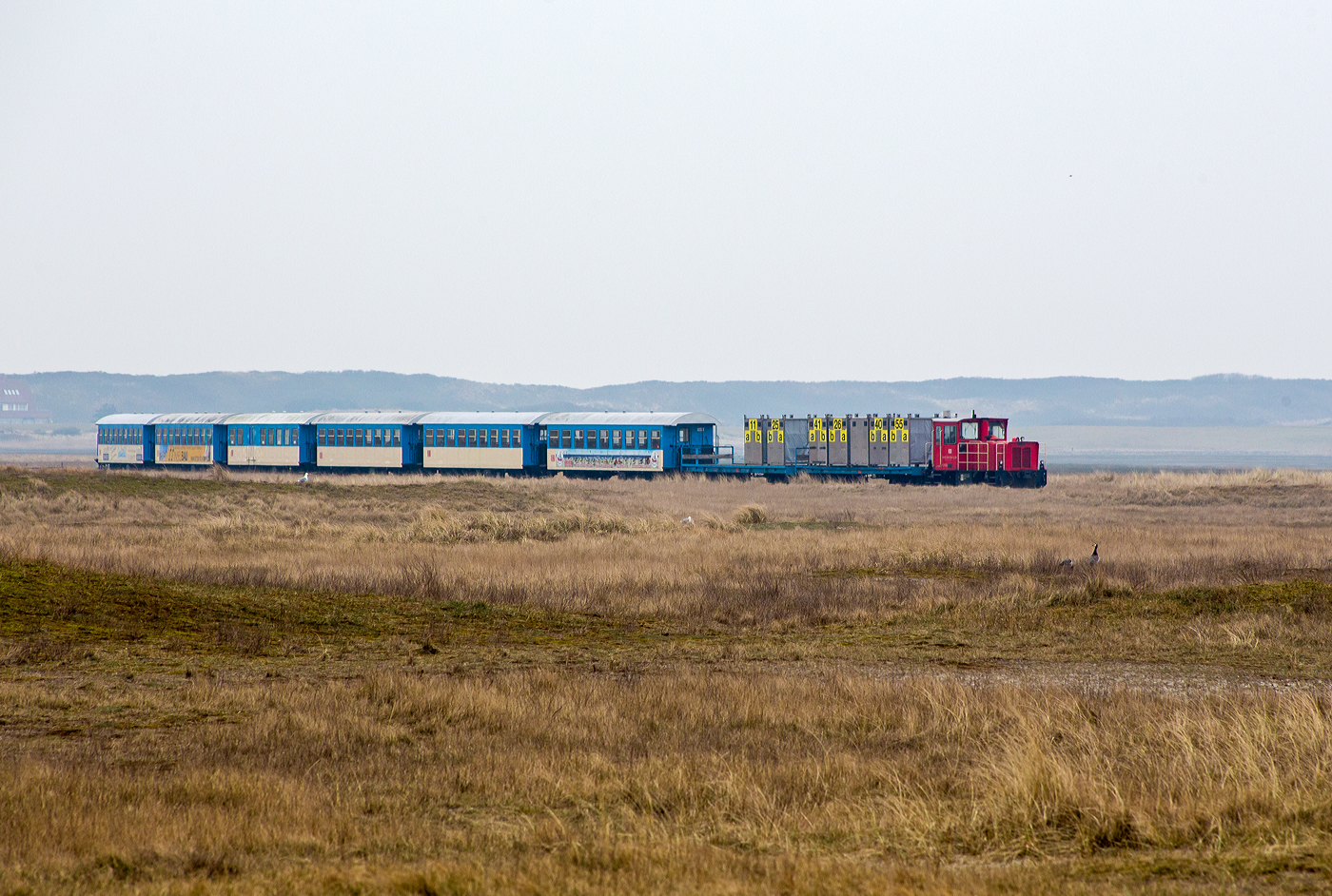Die DB 399 108-0 (98 80 3399 108-0 D-DB), eine meterspurige Schöma CFL150DCL, der DB Fernverkehr AG Standort Wangerooge bzw. der Wangerooger Inselbahn / SIW - Schifffahrt und Inselbahn Wangerooge, kommt mit dem Personenzug und zwei Flachwagen (beladen mit Gepäckcontainern), vom Bahnhof Wangerooge über den Steindamm durch die Salzwiesen und erreicht bald den Westanleger Wangerooge (Fähranleger). 