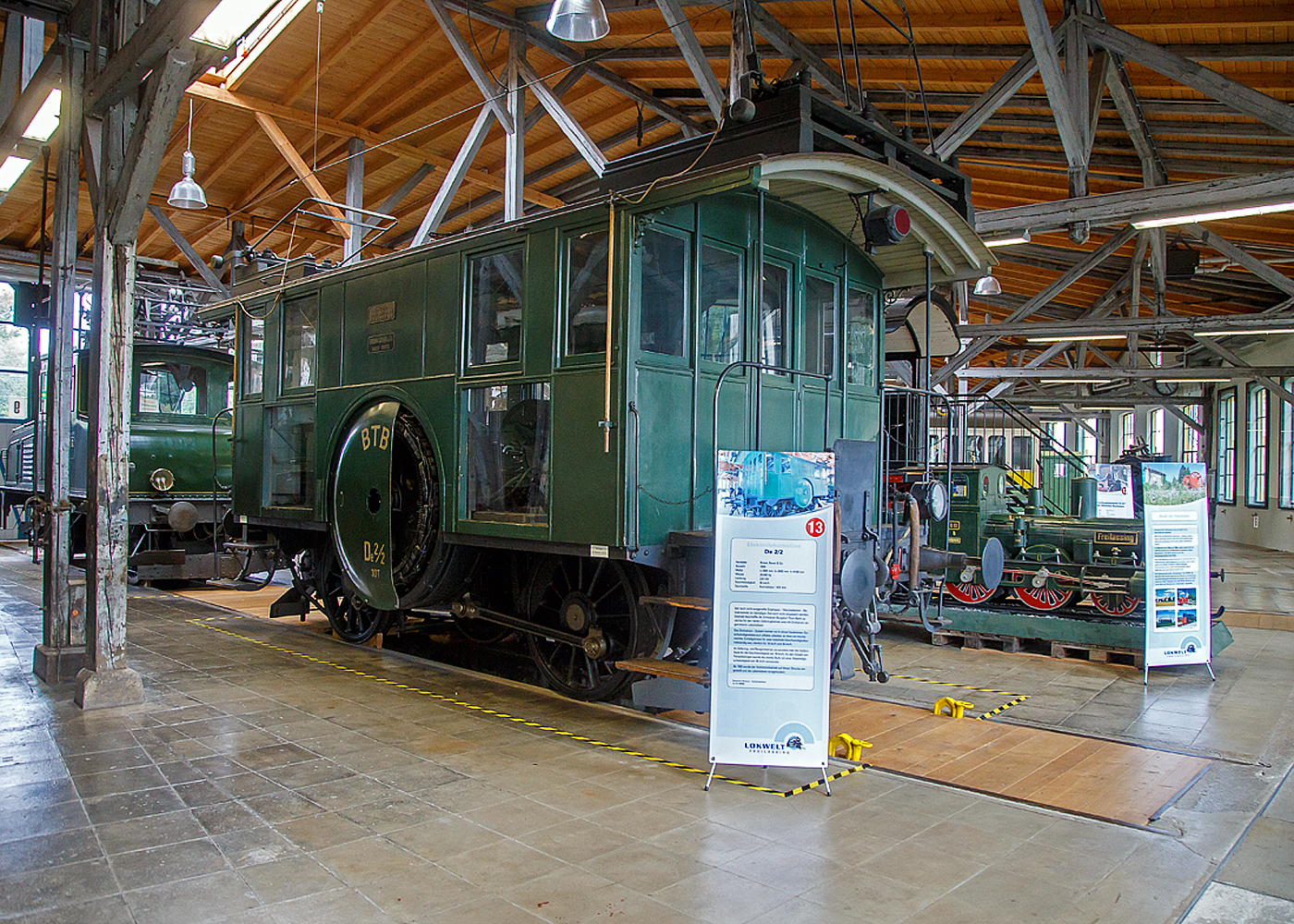 Die Drehstrom- Elektrolokomotive ex BTB De 2/2 Nr.1, ex BTB F 2/2 Nr. 1 (1902 – 1922), ex BTB E2E Nr. 1 (bis 1902), am 11.09.2022 in der Lokwelt Freilassing. Die Lok ist Eigentum des Deutschen Museums - Verkehrszentrum in M�nchen und eine Leihgabe die Lokwelt Freilassing

Dem G�terverkehr der Burgdorf-Thun-Bahn (BTB) dienten zwei Lokomotiven vom Typ F 2/2. Bis 1902 wurden sie als E2E bezeichnet, ab 1922 als De 2/2. Diese beiden ersten in der Schweiz, im Jahr 1899, gebauten elektrischen Lokomotiven (De 2/2 Nr.1 und  2) waren eine gemeinsame Konstruktion der Schweizerischen Lokomotiv- und Maschinenfabrik (SLM) in Winterthur f�r den mechanischen Teil und der Brown, Boveri & Cie. (BBC) f�r die elektrische Ausr�stung. 

Der noch nicht ausgereifte Einphasen-Wechselstrom-Betrieb konnte zur damaligen Zeit noch nicht eingesetzt werden. Deshalb beschaffte die Schweizer Burgdorf-Thun-Bahn (BTB) zun�chst f�r den reinen G�terzugbetrieb zwei dieser mit Drehstrom (Dreiphasenwechselstrom) angetriebenen Lokomotiven. Das Drehstrom-System konnte nur in einem bestimmten Geschwindigkeitsbereich effektiv arbeiten, so dass ein mechanisches Schaltgetriebe f�r zwei maximale Geschwindigkeiten notwendig war, n�mlich f�r 18 und 36 km/h.

Ab 1933 wurde der Drehstrombetrieb auf dieser Strecke eingestellt und die beiden Lokomotiven ausgemustert. Die beiden Lokomotiven sind als weltweit erste Drehstromlokomotiven f�r den Vollbahnbetrieb erhalten geblieben. Die Nr. 1 befindet in der Lokwelt Freilassing, die Nr. 2 befindet sich im Verkehrshaus der Schweiz in Luzern.

Die beiden Motoren wiesen eine Leistung von je 150 PS (110 kW) auf. Das nur im Stillstand umschaltbare Getriebe erm�glichte zwei Geschwindigkeiten, Die eine bis 18 km/h lie� auf der gr��ten Steigung von 25 ‰ eine Anh�ngelast von 100 Tonnen zu, die andere bis 36 km/h noch die H�lfte. Wobei die zul�ssige H�chstgeschwindigkeit betrug 50 km/h, so wurde in den sp�teren Betriebsjahren in Gef�llen stromlos gefahren, um diese zu erreichen. Dank der h�heren Geschwindigkeit h�tten die Lokomotiven n�tigenfalls auch im Personenverkehr Verwendung finden k�nnen.

Die beiden 19-poligen Triebmotoren sind beidseits auf einer auf einem Hilfsgestell gelagerten gemeinsamen Welle montiert. �ber ein Vorgelege wurde das Drehmoment mittels Kuppelstangen auf die beiden Achsen �bertragen. Zum Anfahren war ein f�r beide Motoren gemeinsamer Widerstand vorhanden, der mit dem in jedem F�hrerstand vorhandenen Kontroller allm�hlich ausgeschaltet wurde. Beleuchtung, Heizung und Hilfsbetriebe wurden mit einer Spannung von 100 Volt betrieben. Die Lokomotiven weisen an beiden Enden offene Plattformen auf. Die vier Schleifb�gel auf dem Dach legten sich selbstt�tig beim Wechsel der Fahrtrichtung nach hinten um. Zum Abb�geln mussten die an den B�geln angebrachten kurzen Zugseile vom Boden aus mit h�lzernen Stangen heruntergezogen und in Arretierhaken eingeh�ngt werden.

TECHNISCHE DATEN:
Baujahr: 1899, Ausmusterung1930
Spurweite: 1.435 mm (Normalspur)
Achsfolge: B
L�nge �ber Puffer: 7.800  mm
Achsabstand: 3.140 mm
Treibraddurchmesser:  1.230 mm
Dienstgewicht: 29.600 kg
H�chstgeschwindigkeit: 18 km/h / 36 km/h (stromlos bergab 50 km/h)
Stundenleistung: 300 PS (220 kW)
Stundenzugkraft: 4.400 daN
Stromsystem: 750 V, 40 Hz ∆
Anzahl der Fahrmotoren: 2
�bersetzungsstufen: 2 (f�r 18 / 36 km/h)

Die Burgdorf-Thun-Bahn (BTB) war eine Eisenbahngesellschaft in der Schweiz. Ihre Strecke von Burgdorf �ber Konolfingen nach Thun wurde als erste elektrische Vollbahn der Schweiz im Jahr 1899 dem Betrieb �bergeben. Heute ist sie Bestandteil der BLS AG