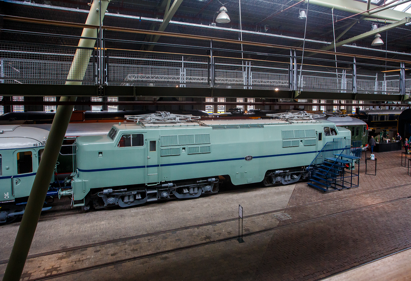 Die Elektrolokomotive NS 1201 der NS-Baureihe 1200 am 24 Juni 2025 im Het Spoorwegmuseum (bis 2005 NSM - Nederlands Spoorwegmuseum / Niederländischen Eisenbahnmuseum) in Utrecht im ehemaligen Bahnhof Maliebaan. Ein Eisenbahnmuseum das zum Verweilen einlädt.

Die Lok wurde 1951 von Werkspoor in Utrecht unter der Fabriknummer 928 gebaut (Baldwin-Lizenz) und an die NS - Nederlandse Spoorwegen (Niederländischen Staatsbahnen, heute Niederländische Eisenbahnen AG) geliefert. Wobei die Entwicklung und Konstruktion, sowie auch einige Teile, von Baldwin Locomotive Works in Philadelphia (USA) stammt. Auch die Entwicklung der Elektrotechnik stammt aus den Vereinigten Staaten und zwar von Westinghouse Electric Corporation (Monroeville), diese wurde von Heemaf in Hengelo (Westinghouse-Lizenz) geliefert.

Die Baureihe 1200 der Niederländischen Eisenbahnen (NS) ist eine sechsachsige Elektrolokomotivbaureihe aus den Jahren 1951 bis 1953. Die Lokomotiven waren bis 1998 bei der NS im Einsatz.

Die Nase war der erste Teil dieser unglaublich starken und robusten Lokomotive, der ins Auge fiel. Sie sollte dem Lokführer bei Kollisionen einen besseren Schutz bieten. Diese Lokomotiven wurden in den Niederlanden nach einem amerikanischen Design und mit einigen amerikanischen Teilen gebaut, die teilweise durch amerikanische Dollar über den Marshall-Plan finanziert wurden. Die NS nutzte diese Lokomotiven, um sowohl Personenzüge als auch Güterzüge zu ziehen. Ihre wichtigste Aufgabe war es Schnellzüge zu ziehen. Als sich die NS 1995 trennte, wurden alle 1200er Jahre an NS Cargo übertragen und begannen im Güterverkehr zu arbeiten. 

Nach Ende des Zweiten Weltkrieges gab es großen Fahrzeugmangel bei der NS. Da man keine Erfahrungen mit dem Bau elektrischer Lokomotiven hatte, kaufte man Lokomotiven bei verschiedenen Lieferanten. Die bei Baldwin konstruierten Lokomotiven wurden bei Werkspoor in Utrecht gefertigt. Die bei Westinghouse entwickelte Elektrotechnik wurde von Heemaf in Hengelo geliefert. Von den ursprünglich 75 georderten Lokomotiven wurden nur 25 ausgeliefert. Der amerikanische Einfluss ist an den hochliegenden Führerständen und den großen, dem Schutz des Lokführers dienenden Vorbauten erkennbar. Am 30. November 1950 wurden 25 Loks bestellt, am 27. September 1951 konnte die erste Probefahrt stattfinden. Am 9. Februar 1952 wurde die 1201 offiziell in Dienst gestellt, der Preis pro Lok betrug 765.000 NFL (ca. 347.000 Euro).

Die Loks hatten sechs Motoren des Typs TM94. Die ersten 14 Lokomotiven waren türkis lackiert, danach setzte sich wegen des sichtbaren Abriebs der Stromabnehmer eine rotbraune Farbgebung durch. Ab 1954 erhielten alle Loks eine blaue Farbgebung, 1971 erhielt die erste Lok die neuen NS-Hausfarben grau/gelb, 1983 war auch die letzte Lok entsprechend umlackiert. Am 30. März 1998 endete der planmäßige Einsatz bei der NS.

Fünf weitere Lokomotiven (1214, 1215, 1218, 1221, 1225) wurden 1999 an die ACTS Nederland BV verkauft. Diese vermietete 2010 drei Lokomotiven (1214, 1215, 1225) an die Eisenbahngesellschaft Euro-Express Treincharter (EETC). Von der wurden sie bis April 2015 unter anderem vor den Nachtzügen Niederlande–Österreich zwischen Amsterdam und Venlo eingesetzt. Fünf Maschinen (1201, 1202, 1211, 1218, 1221) sind als Museumslokomotiven erhalten. NS 1211 ist Botschafterlok im Bahnpark Augsburg. Leihgeber ist das Eisenbahnmuseum Utrecht.

TECHNISCHE DATEN:
Hersteller mech.: Werkspoor (in Lizenz von Baldwin)
Hersteller Elektrik: Heemaf (in Lizenz von Westinghouse)
Baujahre: 1950 bis 1953
Ausmusterung bei NS: bis 1998 
Gebaute Anzahl: 25
Nummerierung: NS 1201 bis 1225
Spurweite: 1.435 mm (Normalspur)
Achsfolge: Co’Co’ 
Länge über Puffer: 18.086 mm
Höhe: 4.064 mm
Treibraddurchmesser: 1.100 mm (neu)
Dienstgewicht: 108 t
Antriebsmotoren: 6 Stück Heemaf vom Typ TM 94
Getriebeübersetzung: 20 : 71
Höchstgeschwindigkeit: 130 km/h (Planmäßig) / 150 km/h (technisch möglich)
Stundenleistung: 6 x 392 kW = 2.350 kW
Dauerleistung: 6 x 367 kW = 2.200 kW
Anfahrzugkraft: 195 kN
Dauerzugkraft: 164 kN bei 80 km/h / 64,7 kN bei 123 km/h
Stromsystem: 1,5 kV DC (=)
Kleister befahrbarer Gleisbogen: R 100 m