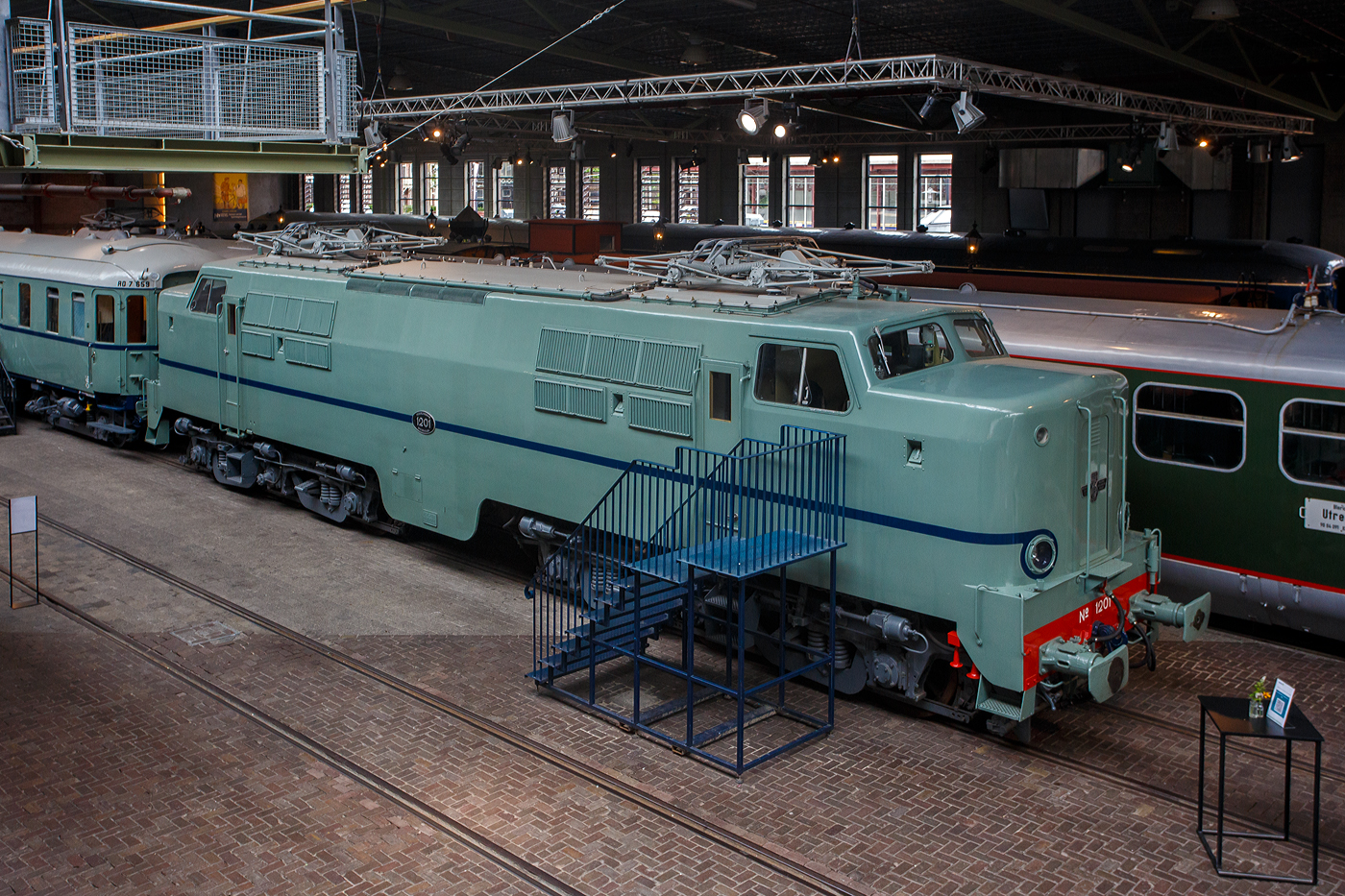 Die Elektrolokomotive NS 1201 der NS-Baureihe 1200 am 24 Juni 2025 im Het Spoorwegmuseum (bis 2005 NSM - Nederlands Spoorwegmuseum / Niederländischen Eisenbahnmuseum) in Utrecht im ehemaligen Bahnhof Maliebaan. Ein Eisenbahnmuseum das zum Verweilen einlädt.

Die Lok wurde 1951 von Werkspoor in Utrecht unter der Fabriknummer 928 gebaut (Baldwin-Lizenz) und an die NS - Nederlandse Spoorwegen (Niederländischen Staatsbahnen, heute Niederländische Eisenbahnen AG) geliefert. Wobei die Entwicklung und Konstruktion, sowie auch einige Teile, von Baldwin Locomotive Works in Philadelphia (USA) stammt. Auch die Entwicklung der Elektrotechnik stammt aus den Vereinigten Staaten und zwar von Westinghouse Electric Corporation (Monroeville), diese wurde von Heemaf in Hengelo (Westinghouse-Lizenz) geliefert.

Die Baureihe 1200 der Niederländischen Eisenbahnen (NS) ist eine sechsachsige Elektrolokomotivbaureihe aus den Jahren 1951 bis 1953. Die Lokomotiven waren bis 1998 bei der NS im Einsatz.

Die Nase war der erste Teil dieser unglaublich starken und robusten Lokomotive, der ins Auge fiel. Sie sollte dem Lokführer bei Kollisionen einen besseren Schutz bieten. Diese Lokomotiven wurden in den Niederlanden nach einem amerikanischen Design und mit einigen amerikanischen Teilen gebaut, die teilweise durch amerikanische Dollar über den Marshall-Plan finanziert wurden. Die NS nutzte diese Lokomotiven, um sowohl Personenzüge als auch Güterzüge zu ziehen. Ihre wichtigste Aufgabe war es Schnellzüge zu ziehen. Als sich die NS 1995 trennte, wurden alle 1200er Jahre an NS Cargo übertragen und begannen im Güterverkehr zu arbeiten. 

Nach Ende des Zweiten Weltkrieges gab es großen Fahrzeugmangel bei der NS. Da man keine Erfahrungen mit dem Bau elektrischer Lokomotiven hatte, kaufte man Lokomotiven bei verschiedenen Lieferanten. Die bei Baldwin konstruierten Lokomotiven wurden bei Werkspoor in Utrecht gefertigt. Die bei Westinghouse entwickelte Elektrotechnik wurde von Heemaf in Hengelo geliefert. Von den ursprünglich 75 georderten Lokomotiven wurden nur 25 ausgeliefert. Der amerikanische Einfluss ist an den hochliegenden Führerständen und den großen, dem Schutz des Lokführers dienenden Vorbauten erkennbar. Am 30. November 1950 wurden 25 Loks bestellt, am 27. September 1951 konnte die erste Probefahrt stattfinden. Am 9. Februar 1952 wurde die 1201 offiziell in Dienst gestellt, der Preis pro Lok betrug 765.000 NFL (ca. 347.000 Euro).

Die Loks hatten sechs Motoren des Typs TM94. Die ersten 14 Lokomotiven waren türkis lackiert, danach setzte sich wegen des sichtbaren Abriebs der Stromabnehmer eine rotbraune Farbgebung durch. Ab 1954 erhielten alle Loks eine blaue Farbgebung, 1971 erhielt die erste Lok die neuen NS-Hausfarben grau/gelb, 1983 war auch die letzte Lok entsprechend umlackiert. Am 30. März 1998 endete der planmäßige Einsatz bei der NS.

Fünf weitere Lokomotiven (1214, 1215, 1218, 1221, 1225) wurden 1999 an die ACTS Nederland BV verkauft. Diese vermietete 2010 drei Lokomotiven (1214, 1215, 1225) an die Eisenbahngesellschaft Euro-Express Treincharter (EETC). Von der wurden sie bis April 2015 unter anderem vor den Nachtzügen Niederlande–Österreich zwischen Amsterdam und Venlo eingesetzt. Fünf Maschinen (1201, 1202, 1211, 1218, 1221) sind als Museumslokomotiven erhalten. NS 1211 ist Botschafterlok im Bahnpark Augsburg. Leihgeber ist das Eisenbahnmuseum Utrecht.

TECHNISCHE DATEN:
Hersteller mech.: Werkspoor (in Lizenz von Baldwin)
Hersteller Elektrik: Heemaf (in Lizenz von Westinghouse)
Baujahre: 1950 bis 1953
Ausmusterung bei NS: bis 1998 
Gebaute Anzahl: 25
Nummerierung: NS 1201 bis 1225
Spurweite: 1.435 mm (Normalspur)
Achsfolge: Co’Co’ 
Länge über Puffer: 18.086 mm
Höhe: 4.064 mm
Treibraddurchmesser: 1.100 mm (neu)
Dienstgewicht: 108 t
Antriebsmotoren: 6 Stück Heemaf vom Typ TM 94
Getriebeübersetzung: 20 : 71
Höchstgeschwindigkeit: 130 km/h (Planmäßig) / 150 km/h (technisch möglich)
Stundenleistung: 6 x 392 kW = 2.350 kW
Dauerleistung: 6 x 367 kW = 2.200 kW
Anfahrzugkraft: 195 kN
Dauerzugkraft: 164 kN bei 80 km/h / 64,7 kN bei 123 km/h
Stromsystem: 1,5 kV DC (=)
Kleister befahrbarer Gleisbogen: R 100 m