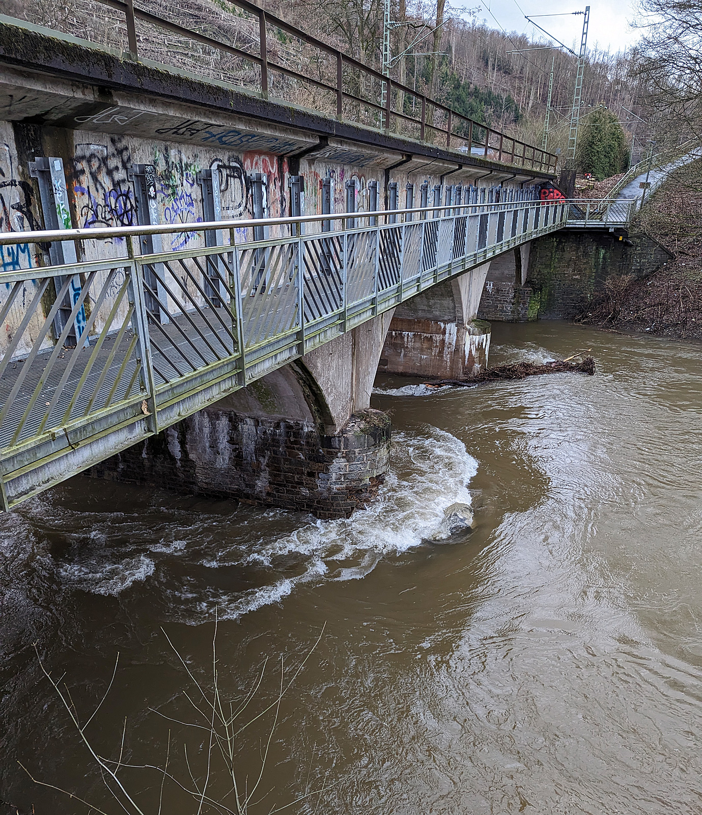Die vom Hochwasser besch�digte Eisenbahn-Siegbr�cke mit Fu�g�ngersteg in Kirchen (Sieg), der Siegstrecke (KBS 460), hier am 03.01.2024 (Bild mit Smartphone).

Die Bahnstrecke Siegen - K�ln wird zwischen Kirchen und Betzdorf bis min. Ende n�chster Woche gesperrt bleiben. Das hat die Deutsche Bahn Dienstagnachmittag mitgeteilt. Durch das Sieg-Hochwasser gibt es Sch�den an einer Eisenbahnbr�cke in Kirchen (kurz vorm Bahnhof). Aufgrund der Wetterlage und der weiterhin anhaltenden Regenf�lle sei die Sperrung erforderlich, so die Bahn-Pressestelle. Die Z�ge des RE 9 k�nnen wegen der Streckensperrung nur bis Betzdorf fahren. Zwischen Siegen und Kirchen verkehrt die Regionalbahn 90 der Hessischen Landesbahn, leider aber nur wenige und teils mit „kleinen“ LINT 27 Triebwagen. Da w�re wahrhaft mehr m�glich und man m�sste den SEV nur zwischen Betzdorf und Kirchen betreiben. Aber aufgrund aktuell hoher Krankenst�nde kommt es auf der Linie RE 9 zudem zu Einschr�nkungen bis 12.01.2024.

Ich konnte mir den Schaden an einen Br�ckenpfeiler selbst anschauen (Bilder folgen) und finde die Streckensperrung richtig, dar�ber lie� ich auch keinen mit Personen besetzten Zug fahren. Was w�re wenn ein Zug dar�ber f�hrt und dadurch einbricht. Wer will das verantworten!!! 

Am 29.12.2023 wollten wir mit der Bahn nach Siegen, durch die Reiseauskunft unter www.bahn.de sahen wir bereits das durch wegen einer besch�digten Br�cke zwischen Betzdorf (Sieg) und Kirchen (Sieg) ist eine Br�cke besch�digt. Die Z�ge der Linie RE9 aus Richtung K�ln enden und beginnen demnach in Betzdorf (Sieg). Die Z�ge der Linie RE 9 aus Richtung Siegen Hbf enden und beginnen demnach in Kirchen, diese waren aber nicht zu sehen. Bis dahin gab es keine Informationen zur Dauer der Sperrung. Somit fuhren wir mit dem Auto nach Kirchen und fuhren dann mit der RB 90 der HLB (Hessischen Landesbahn) in einem LINT 27, wie die �lsardinen, nach Siegen Hbf. Auf der R�ckfahrt um 14:31 Uhr (ab Siegen) mit RB 90 fuhren wir in einem LINT 41.

Am 30.12.2023 fuhren wir nochmal nach Siegen, das Wetter war besser und unser Ticket war noch g�ltig. Die R�ckfahrt von Siegen machten wir um 13:31 Uhr mit dem HLB RB 90 nach Altenkirchen, dem HLB VT 261 einem LINT 41, dieser war der erste Zug der wieder durchfuhr und die Br�cke in langsamer Fahrt befuhr. 

Sp�ter hie� es: Die Br�cke zwischen Betzdorf und Kirchen ist nach wie vor besch�digt. Die Strecke ist aber wieder befahrbar. Die Z�ge fahren in dem betroffenen Streckenabschnitt langsamer. Reisende m�ssen mit Verz�gerungen rechnen und sollten ihre Reiseverbindung kurz vor der Abfahrt des Zuges �berpr�fen.

Nun ist aber der betroffene Streckenabschnitt wieder gesperrt, die Br�cke kann nicht mehr befahren werden.
