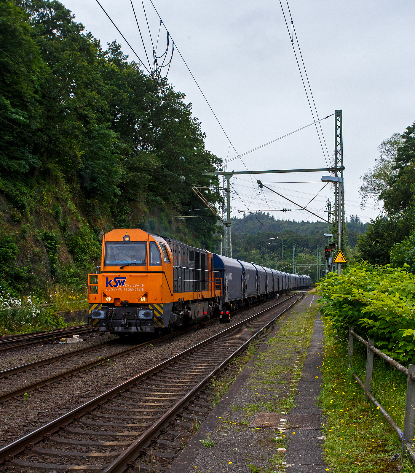 Die KSW 43 (92 80 1273 018-2 D-KSW), die asymmetrische Vossloh G 2000 BB der KSW (Kreisbahn Siegen-Wittgenstein) fährt am 03 Juli 2024, mit einem beladen Coilzug (16 Stück VTG Coilwagen der Gattung Shimmns-ttu, VTG Bez. F46.520D) vom Hafen Duisburg kommend, durch den Bahnhof Scheuerfeld(Sieg) in Richtung Herdorf via Betzdorf. 

In Betzdorf (Sieg) geht es dann über KBS 462 „Hellertalbahn“ nach Herdorf zum Rbf der KSW (Betriebsstätte FGE -Freien Grunder Eisenbahn). Für die Weiterfahrt auf den Pfannenberg (in Neunkirchen-Salchendorf) über nimmt dort dann die KSW 47 (92 80 1271 027-5 D-KSW), ex D 2 der HFM, eine Vossloh G 1000 BB, den Zug.
