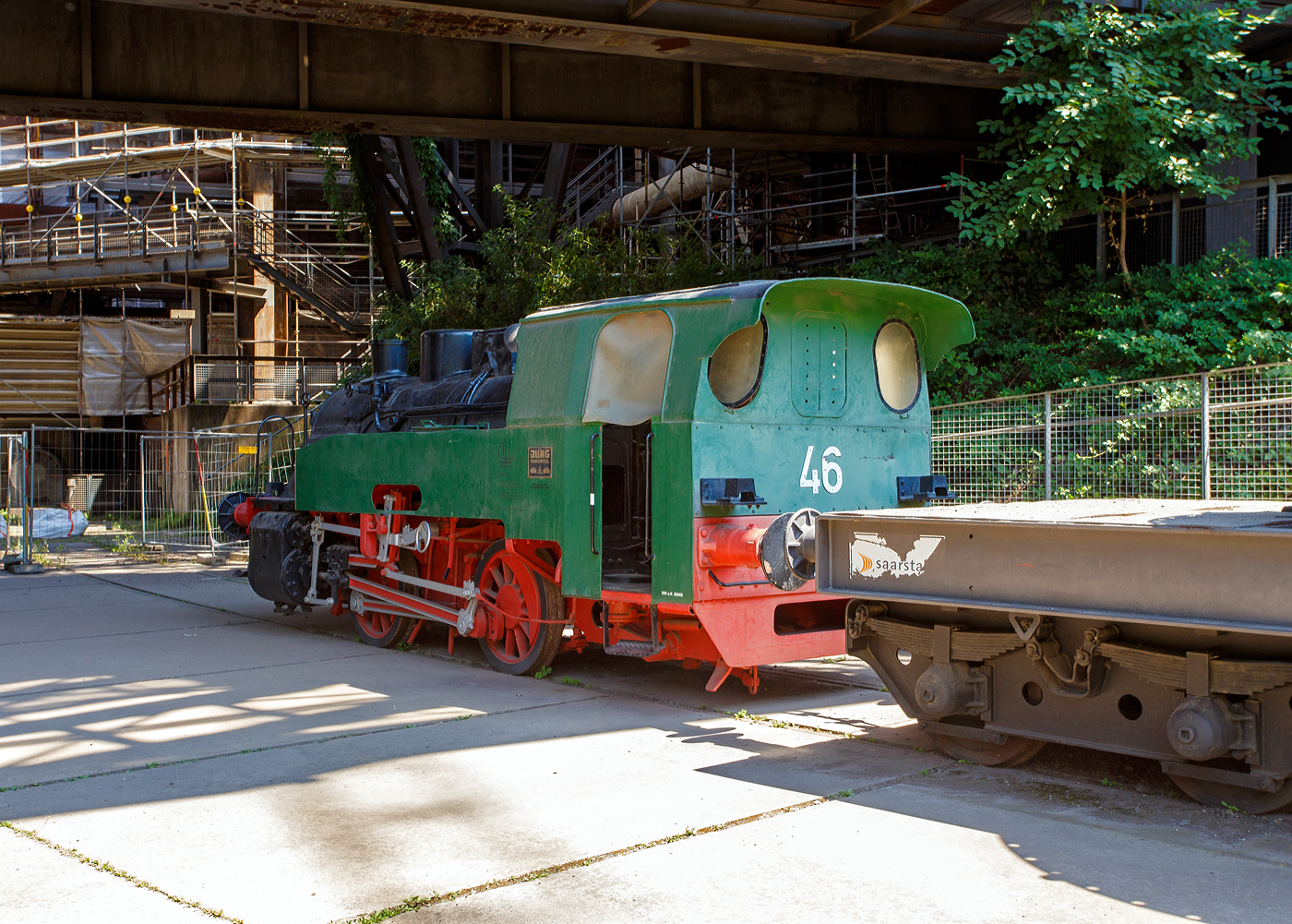 Die Normalspur-Dampflokomotive 46 der V�lklinger H�tte (ehem. R�chling'schen Eisen- und Stahlwerke GmbH in V�lklingen/Saar, ab 1982 ARBED-Saarstahl GmbH, ab 1986 Saarstahl V�lklingen GmbH), eine Jung Hochofenlok niedriger Bauart, abgestellt mit einem offenen Roheisenpfannenwagen der Burbacher H�tte, hier am 01 Juli 2025 am Gel�nde V�lklinger H�tte als Denkmal. Heutiger Eigent�mer ist die Initiative V�lklinger H�tte e.V..

1964 wurde die Lok Nr. 46 mit der Fabriknummer 13835 im Auftrag der R�chling'schen Eisen- und Stahlwerke als letzte rostgefeuerte Dampflok der Firma Arnold Jung, Lokomotivfabrik GmbH, in Jungenthal bei Kirchen an der Sieg gebaut und am 09.12.1964 geliefert. Einsatzgebiet war der Roheisentransport von den sechs Hoch�fen zum Thomasstahlwerk, wobei die besonders beengten Verh�ltnisse auf der V�lklinger H�tte eine besondere Bauform erforderten. Die Verbindung zwischen Eisenwerk und Stahlwerk war nur durch einen Tunnel m�glich, da die Deutsche Bahn durch ihre Gleisanlagen die H�tte in zwei Bereiche teilt. Dieser Tunnel, der Roheisenkanal, ist so niedrig, dass die zweiachsige Lok nur eine H�he von 2.640 mm �ber der Schienenoberkante und eine Breite von 2.450 mm aufweist. Die Loks 44 (11945/Baujahr 1953) und 45 (13240/Baujahr 1959), die nach vorliegenden Unterlagen technisch baugleich sind, wurden schon vorher (von der Arnold Jung GmbH an die V�lklinger H�tte geliefert. 

Um speziell die F�hrerhausr�ckwand, die Lampen, aber auch die Kupplungselemente zwischen Lok und dem ersten Wagen vor fl�ssigen gl�henden Spritzern von herabfallenden Roheisen zu sch�tzen, erhielten die Lokomotiven ein nach hinten verl�ngertes Dach. Die Puffer wurden im Stahlwerk den dort vorhanden Roheisenpfannenwagen angepasst. Alte Jung Mitarbeiter berichten, an der Lok h�tte man kaum was verdient, weil l�nst nicht mehr Dampflokteile ab Lager vorhanden waren umd z.T. m�hsam und teuer beschafft werden musste.

Im Zuge der Modernisierung und Rationalisierung wurden die Roheisendampfloks 1981 nicht mehr ben�tigt. W�hrend die Lokomotiven 44 und 45 an ein Museum im Ruhrgebiet verkauft wurden, gelangte die Lokomotive 46 als technisches Denkmal 1982 zur Universit�t Kaiserslautern. Diese schenkte die Lokomotive 46 der Initiative V�lklinger H�tte, die sie mit Hilfe von Sponsoren am 9. Februar 1999 nach V�lklingen zur�ckholte.

TECHNISCHE DATEN:
Baujahre: 1953,1959 und 1964
Gebaute Anzahl: 3
Spurweite: 	1.435 mm (Normalspur)
Achsformel: B
L�nge: 7.600 mm
Achsabstand: 2.000 mm
Triebraddurchmesser: 1.000 mm (neu)
Breite: 2.450 mm
H�he: 2.650 mm
Dienstgewicht: 26 t
H�chstgeschwindigkeit: k. A. 
Leistung: 250 PS
Zugkraft: 7,5 t
Zylinder: 2 St�ck, � 380 mm x 500 mm Hub
Dampfdruck: 14 bar
Heizfl�che: 58,92 m�
Rostfl�che: 1,17 m�
Anzahl Siederohre: 178 St�ck
Durchmesser Siederohre: 33/38 mm
L�nge Siederohre: 2.540 mm
Wasservorrat: 2.500 l (2,5 m�)
Kohlenvorrat: 500 kg