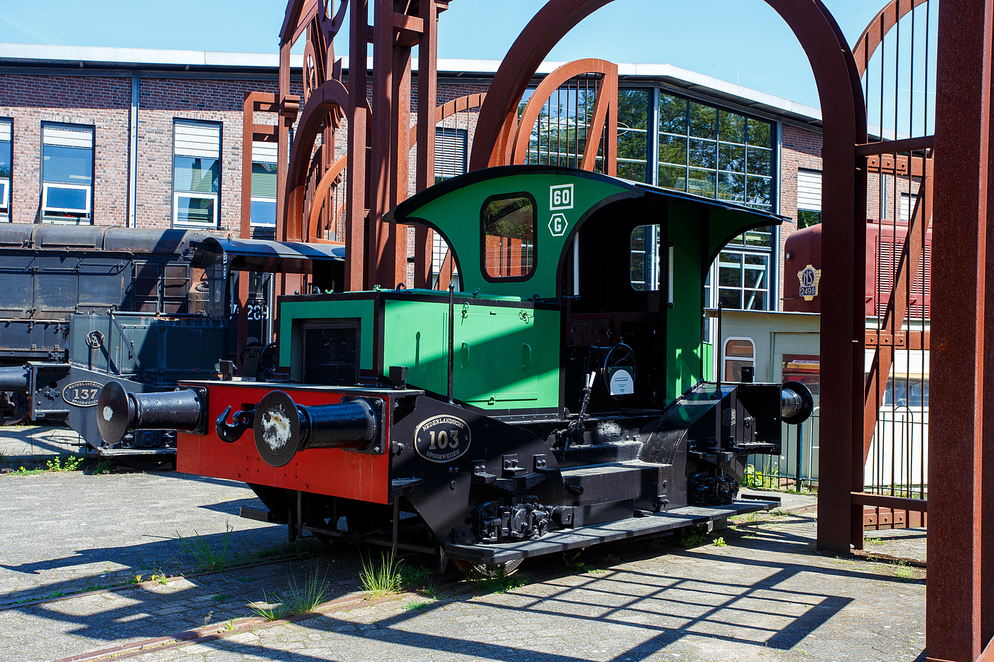 Die Oersik NS 103 Diesel-Rangierlokomotive (ehemals Benzin-Rangierlokomotive) der NS-Serie (Baureihe) 100 am 29 April 2025 im Het Spoorwegmuseum (bis 2005 NSM - Nederlands Spoorwegmuseum / Niederl�ndischen Eisenbahnmuseum) in Utrecht im ehemaligen Bahnhof Maliebaanstation. 

Die NS 103 wurde 1930 von der BMAG - Berliner Maschinenbau-Actien-Gesellschaft vormals L. Schwartzkopff in Berlin unter der Fabriknummer 9734 gebaut und an die NS - Nederlandse Spoorwegen (Niederl�ndischen Staatsbahnen, heute Niederl�ndische Eisenbahnen AG) geliefert. Urspr�nglich hatte sie einen 50 PS starken Benzinmotor von der Heinrich K�mper Motorenfabrik AG in Berlin-Marienfelde (D). Nach dem Krieg wurden die meisten Lokomotiven au�er Betrieb genommen. Dies geschah auch mit der 103 im Jahr 1947 oder 1948, danach wurde sie an NV Staalwerken „De Maas“ in Maastricht verkauft.

Die NS 103 und die NS 137 wurden Ende der 1980er Jahre in die Sammlung des Eisenbahnmuseums aufgenommen. Bei der Revision in Tilburg wurde bei der 103 der Benzinmotor durch einen DAF-Dieselmotor und der mechanische Antrieb durch einen hydrostatischen ersetzt. �u�erlich wurde die Lokomotive wieder in den Originalzustand versetz.

Bei der NS-Lokomotive der Baureihe 100 handelt es sich um benzinbetriebene Lokomotiven, die zwischen 1927 und 1948 bei der Niederl�ndischen Eisenbahn im Einsatz waren. Sie wurden haupts�chlich zum Rangieren und f�r kurze G�terz�ge eingesetzt. Dieser Typ wird auch als Sik oder Oersik bezeichnet.

Die Prototypen 101 und 102:
Im Jahr 1927 wandte sich die NS an die Lokomotivfabrik L. Schwartzkopff in Berlin, die die Lokomotive 101 baute. Die Lokomotive erhielt die Nummer 101 und war eine solide Konstruktion, noch ohne Schutzdach f�r den Lokf�hrer. Zwei Jahre sp�ter (1929) wurde eine weitere Lokomotive gebaut. Dieses hatte einen gr��eren Radstand und ein Schutzdach f�r den Lokf�hrer. Diese Lokomotive verf�gte au�erdem �ber einen st�rkeren 50-PS-Benzinmotor. 

Nach diesen zwei Prototyp-Rangierfahrzeugen, wurde eine Serie von 17 Exemplaren bei der Berliner Maschinenbau A.G. in Berlin bestellt, da man mit der 102 zufrieden war. Diese wurden 1930 gebaut. Ein Jahr sp�ter wurden weitere 11 Einheiten gebaut, diesmal von Werkspoor in Amsterdam. Im Jahr 1932 baute dieses Unternehmen weitere 22 Lokomotiven, die �ber einen st�rkeren Motor (65 PS) verf�gten. Nach der Einf�hrung der neueren Lokomotiven der Serien 200 und 300 wurden diese Lokomotiven seltener eingesetzt. In den letzten Jahren wurden sie vor allem f�r saisonale Rangierarbeiten, beispielsweise zum Verschieben von R�benwaggons, eingesetzt. 1948 verlie� der letzte die NS.

Die NS 103 bis 152 wurden in den 1930er Jahren als billigere Option f�r die Verj�ngung an kleineren Bahnh�fen gebaut. Das Problem war, dass der Einsatz von Dampflokomotiven zum Rangieren von G�terwagen extrem teuer war. Sie mussten stundenlang angeheizt werden und waren gesetzlich verpflichtet, eine Besatzung von mindestens zwei Personen (Lokf�hrer und Heizer) zu haben, wenn sie sich bewegten. Die NS-L�sung bestand darin, eine Reihe von Lokmotoren aus Deutschland zu bestellen. Diese Serie, deren Zahl 103 bis 152 zugewiesen wurde, umfasste einfache Lokomotiven mit einem Benzinmotor (und damit begrenzter Zugkraft), mechanische Bremsen und einer „automatischen“ Kupplung zum Anbringen von G�terwagen. Sie waren relativ preiswert und einfach zu bedienen. Eine Lok war fast sofort einsatzf�hig. Jeder Kleinlokf�hrer, der eine kurze Ausbildung durchlaufen hat, kann sie sicher bedienen. Der letzte dieser „Original-Loks“ wurde 1948 bei der NS aus dem Dienst ausgemustert, woraufhin viele der Lok ein zweites Leben in privaten Industriebahnen begannen. 

Die NS 100 wurde fr�her auch als Assistent einer gro�en Rangierlokomotive auf einem gr��eren Rangierbahnhof eingesetzt. Die Aufgabe des Sik bestand darin, die rangierten Wagengruppen zusammenzudr�cken, damit sie gekuppelt werden konnten. Das Rangieren mit diesen Lokomotiven war aufgrund ihrer geringen Zug- und vor allem Bremskraft nicht schnell. Das Ziehen oder Schieben von mehr als sechs beladenen G�terwagen wurde schnell problematisch. Die Lokomotive hatte keine Druckluftbremse und war anfangs mit einer automatischen Kupplung ausgestattet. Eine Bedienung der Lokomotive war auch �ber die seitlichen Trittbretter m�glich.

Die Lokomotiven waren mit einem Benzinmotor von der Heinrich K�mper Motorenfabrik AG in Berlin–Marienfelde(D) ausgestattet, der �ber eine mechanische Kupplung mit Reibscheiben und Rollenketten beide Achsen antreibt. Die Anschaffung und Wartung einer teuren Dampflok war nicht mehr notwendig, da die Lokomotive relativ g�nstig in der Anschaffung ist und jederzeit gestartet und eingesetzt werden kann. Um gesetzliche Vorschriften zu umgehen, wurde das Fahrzeug nicht Lokomotive, sondern Lokomotor (Kleinlok) genannt. Dadurch und aufgrund der �u�erst einfachen Bedienung konnte die Lokomotive nach einer kurzen Einweisung von einem Rangierf�hrer bedient werden, statt von einem teuren Vollblut-Lokf�hrer und seinem zugeh�rigen Heizer.

TECHNISCHE DATEN:
Hersteller: Berliner Maschinenbau AG (D) / Werkspoor, Amsterdam NL
Benzinmotorhersteller: H. K�mper Motorenfabrik AG, Berlin–Marienfelde(D)
Gebaute Anzahl: 52 (NS 101, 102, 103-152)
Baujahre: Serie ab 1930 bis 1932
Ausmusterung: 1948
Spurweite: 1.435 mm (Normalspur)
Achsfolge: A-A
L�nge �ber Puffer: 5.780 mm (101- 5.000 mm)
Achsabstand: 2.464 bis 2.524 mm (101 – 2.200 mm / 102 – 2.500 mm)
Dienstgewicht: 12,5 t (NS 101 - 8,5 t)
Leistung: 50 PS, sp�tere 65 PS (101 – 20 PS / 102 – 30 PS)
Leistungs�bertragung: mechanisch �ber Rollenketten auf beide Achsen
H�chstgeschwindigkeit: 30 km/h (NS 101 - 10 km/h)


