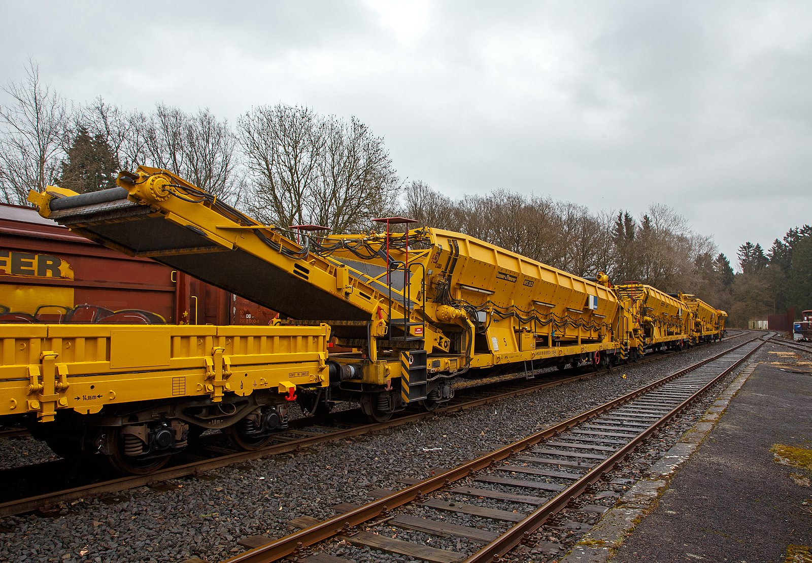 Die Plasser & Theurer Materialförder- und Siloeinheiten MFS 38-D (teilweise auch BSW 2000 - Bunker-Schüttgut-Wagen genannt) der HERING Bahnbau (Burbach), Nr. 5, Schweres Nebenfahrzeug Nr. D-HGUI 99 80 9552 124-4, ex 97 19 22 582-4, ist am 13.03.2023 auf dem Betriebshof der Westerwaldbahn (WEBA) auf der Bindweide bei Steinebach/Sieg abgestellt.

Diese MFS 38-D wurde 2002 von Plasser & Theurer unter der Fabriknummer 4035 gebaut. Die vierachsigen Fahrzeugrahmen und Drehgestelle sind moderne als die Wagen Nr. 1, 3 und 4 der Hering Bau.

Bei diesem MFS 38-D (das „D“ bedeutet Durchförderer) ist das Übergabeband starr (nicht schwenkbar), so ist ein  Entladen nebens Gleis nicht möglich. Die MSF 38-D haben ein Fassungsvermögen 38 m³ bzw. eine Nutzlast von 41,5 t, bei einem Eigengewicht von 38,5 t. Die Förderbänder werden hydraulisch angetrieben. Der Antrieb des Hydraulikaggregates erfolgt durch einen luftgekühlten Deutz Dieselmotor vom Typ F6L 912 mit einer Leistung von 82 kW.

TECHNISCHE DATEN:
Maschinenart: Material-Förder-Siloeinheit MSF 38
Hersteller: Plasser & Theurer
Spurweite: 1.435 mm (Normalspur)
Anzahl der Achsen: 4 (in 2 Drehgestellen)
Länge über Puffer:19.900 mm
Gesamtlänge mit Übergabeband: 23.550 mm
Drehzapfenabstand: 14.860 mm
Achsabstand im Drehgestell: 1.800 mm
Breite: 2.890 mm
Höhe: 4.280 mm
Eigengewicht: 38,5 t
Max. Zuladung (Nutzlast): 41,5 t
Ladevolumen: 38 m³
Höchstgeschwindigkeit: 100 km/h (geschleppt)
Kleister befahrbarer Gleisbogen: R 35 m
Bremse: KE – GP - A
Motor: luftgekühlter Deutz Dieselmotor vom Typ F6L 912 F
Leistung: 82 kW
Dieseltankvolumen: 750 l
Übergabeband: starr
Die Förderbänder sind in folgenden Geschwindigkeiten regelbar:
Siloband: 1. Gang (Speichern)   0 - 0,028 m/s / 2. Gang (Durchfördern)  0 - 0,140 m/s
Übergabeband: 0 - 1,000 m/s

Einsatz und Arbeitsweise:
Mit den Materialförder- und Siloeinheiten können unterschiedliche Schüttgüter wie beispielsweise Abraum, Planum- und Frostschutzmittel oder Bettungsstoffe unkompliziert, geladen, gefördert, gespeichert und entladen werden. Das Abraumverladesystem ist weitestgehend automatisiert und benötigt zur Bedienung nur wenig Personal. Das System verfügt zudem über eine autarke Energieversorgung (durch Dieselmotore). Ein unabhängiges Entladen ist durch schwenkbare Übergabebänder an jeder geeigneten Stelle möglich. Die MFS-Wagen können in Kombination mit einer Beladestation in beliebiger Anzahl aufgereiht und vor Kopf be- und entladen werden.

Am Boden jedes MFS 38 befindet sich ein über die ganze Wagenbreite gehender Fördergurt und an der Stirnseite ein breites Abgabeförderband. Das Aushubmaterial fällt von der Maschine kommend auf den Bodengurt, wird zum Abgabeförderband gebracht und an die nächste Einheit übergeben. Dieser Vorgang wiederholt sich, bis die am weitesten entfernte Siloeinheit erreicht wird. Bei dieser hintersten Siloeinheit ist das Abgabeförderband an der Stirnseite des Wagens in Ruhestellung. Auch der Fördergurt am Boden des Wagens ist so lange in Ruhestellung, bis die obere Füllgrenze erreicht ist. Dann wird der Fördergurt in Bewegung versetzt und dadurch im Übergabebereich freier Raum für die weitere Beladung geschaffen. Durch die dem Fülltempo angepasste Geschwindigkeit des Fördergurts wird die ganze Siloeinheit vollständig gefüllt. Das Fassungsvermögen einer Siloeinheit beträgt 38 Kubikmeter.

Nach Füllung einer Teilgruppe des Abraumzuges kann diese von der Restgruppe abgekuppelt werden, zu einer Entladestelle gefahren und dort entleert werden, während die Restgruppe des Abraumzuges noch befüllt wird. Das Entladeband ist nach beiden Seiten ausschwenkbar. Die Entladung kann dabei auf einer Deponie erfolgen oder es können LKW's oder Waggons am Nachbargleis beladen werden. Die MFS 38 (ohne nachfolgendes „D“) des Abraumzuges können durch Ausschwenken aller Abgabeförderbänder gleichzeitig entleert werden. Die Entladung eines Wagens dauert ungefähr 3,5 Minuten.
