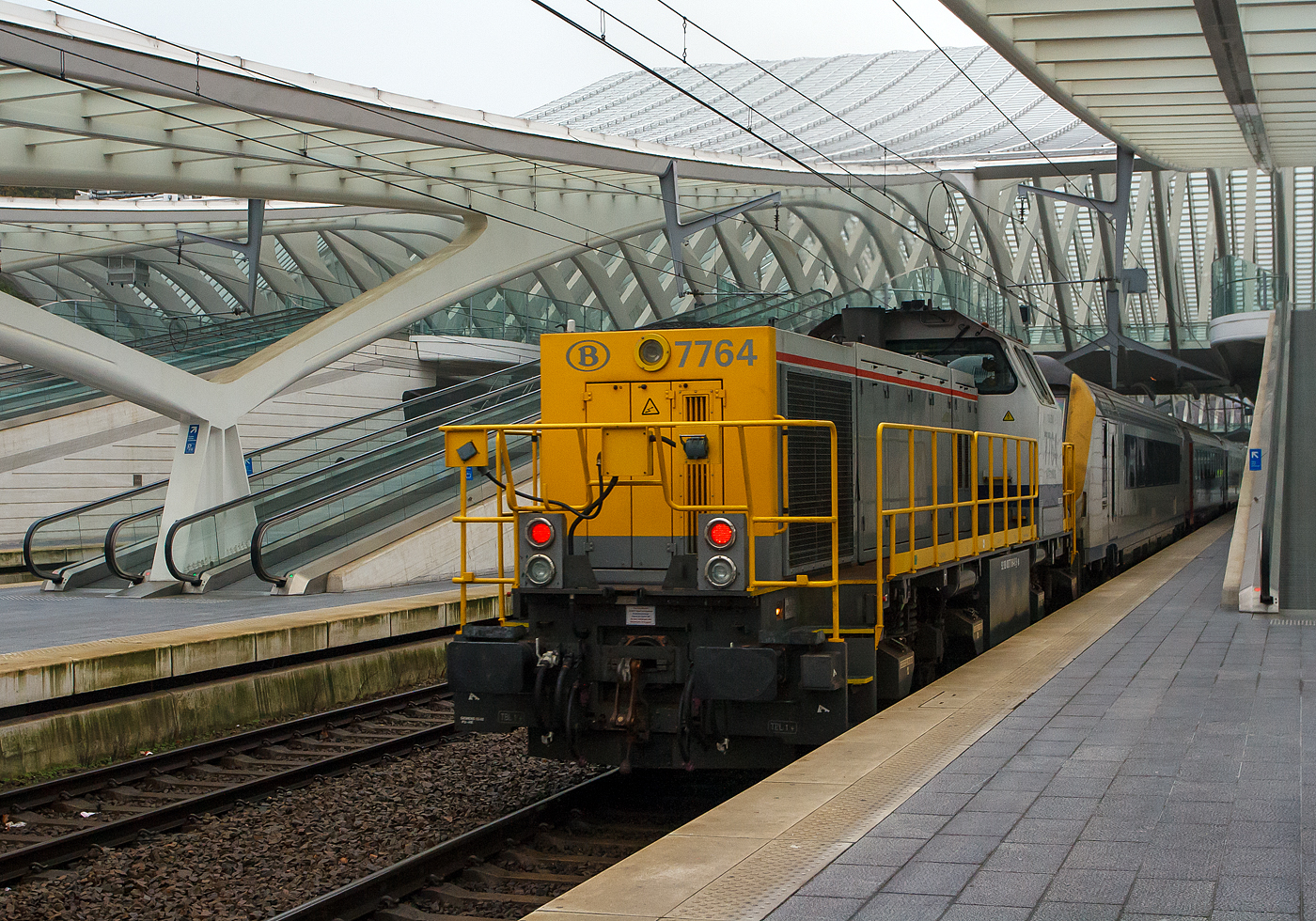 Die SNCB/NMBS HLD 7764  Toledo  leistet am 23 November 2013, einem IC nach Ostende, vom Bahnhof Lüttch-Guillemins, Schubhilfe.

Nach der Ausfahrt vom Bahnhof Lüttch-Guillemins (Gare de Liège-Guillemins) in Richtung Brüssel, der Bahnstrecke Brüssel – Lüttich (Linie 36) folgt bald die 30 ‰ steile Rampe Haut Pré, darum muss bei langen schweren Zügen eine Lok Schubhilfe leisten. Früher war oben am Haut Pré die Bergstation einer Kabelzuganlage. Hier kommt die SNCB/NMBS HLD 7764  Toledo  am 23.11.2013 nach getaner Arbeit (Schubhilfe) retour. 

Die Lok wurde 2002 unter der Fabriknummer 1000981 bei Vossloh in Kiel gebaut, sie trägt die NVR-Nummer: 92 88 0077 064-8 B-B. 

Die HLD77 ist eine speziell auf die Belange der Belgischen Staatsbahn (SNCB) zugeschnittene Weiterentwicklung der G 1205. Zwischen 1999 und 2005 wurden insgesamt 170 Lokomotiven an die SNCB geliefert. Neben dem belgischen Dieselmotor, einen Anglo-Belgian Cooperation 6-Zylinder-Dieselmotor vom Typ 6DZC-1000-144A mit 1.150 KW Leistung, und dem elektrischen Hilfsbetriebekonzept ohne Hydrostatikanlage unterscheidet sie sich in zahlreichen Details von den Standardloks, womit den speziellen Wünschen des Kunden Rechnung getragen wurde. Das Turbowendegetriebe verfügt über ein im Stillstand schaltbares Stufengetriebe für zwei Geschwindigkeitsbereiche. Die Leistungsübertragung vom Getriebe geschieht über Kardanwellen (Gelenkwellen) auf alle 4 Achsen.

Die Lokomotiven kommen im Rangier- und Streckendienst zum Einsatz. Die Lokomotiven 7771 bis 7790 sind zusätzlich in Deutschland und den Niederlanden zugelassen und mit den entsprechenden Zugsicherungen ausgerüstet.

TECHNISCHE DATEN:
Baujahre: 1999 - 2005
gebaute Stückzahl: 170
Spurweite: 1.435 mm (Normalspur)
Achsfolge: B´B´
Länge über Puffer: 15.590 mm
Drehzapfenabstand: 7.770 mm
Achsabstand im Drehgestell: 2.400 mm
größte Breite: 3.052 mm
größte Höhe über Schienenoberkante: 4.220 mm
kleinster befahrbarer Gleisbogen: R 75 m
Dienstgewicht: 90 t

Motordaten:
Dieselmotorhersteller: Anglo-Belgian Cooperation AG (ABC), Gent (Belgien)
Motorart: Wassergekühlter Mittelschnellläufer Viertakt-6-Zylinder-Reihendieselmotor mit Turboaufladung und Ladeluftkühlung (Wassergekühl), sowie Direkteinspritzung (eine Pumpe/Zylinder)
Motortyp: 6(T)DZC-1000-144A 
Motorleistung: 1.150 kW (1.536 PS)
Motordrehzahl: 1.000 U/min
Hubraum: 95,7 Liter (Bohrung Ø 256 mm x 310 mm Hub)

Getriebe: Voith L4r4zseU2a
Hilfsgenerator, dreiphasig: 65–130 kW
Höchstgeschwindigkeit: 60 km/h / 100 km/h (Rangier-/Streckengang)
Anfahrzugkraft: 265 / 210 kN (Rangier-/Streckengang)
Kraftstoffvorrat: 4.200 l
kleinster befahrbarer Gleisbogen: R 75 m
