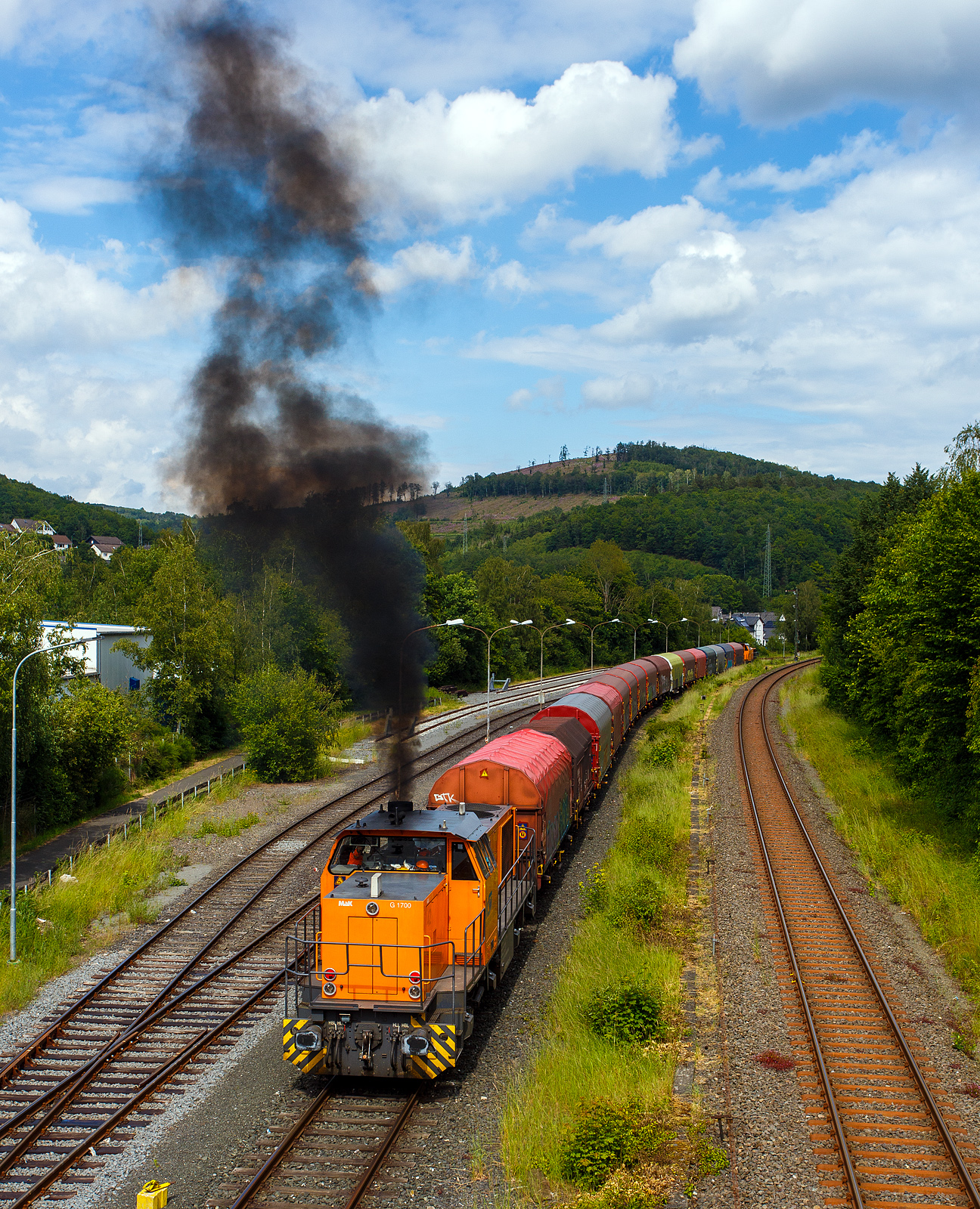 Dieselwolken im Hellertal, die KSW 42 (92 80 1277 902-3 D-KSW), eine Vossloh MaK G 1700 BB, drückt den schweren Coilzug und ganz vorne zieht die KSW 46 bzw. 277 807-4 (92 80 1277 807-4 D-KSW), eine Vossloh G 1700-2 BB, vom KSW Rangierbahnhof Herdorf (Betriebsstätte Freien Grunder Eisenbahn - NE 447) nun in Richtung Neunkirchen-Salchendorf (ehemals Pfannenberger Einigkeit). Beide Loks gehören der KSW (Kreisbahn Siegen-Wittgenstein), die wie hier werktäglich (ehemals Pfannenberger Einigkeit) ihre Coilzüge zu den Produktionshallen der EMW Stahl Service GmbH, ein Geschäftsbereich der SCHÄFER WERKE Gruppe befördern. Schon heute werden so über 300.000 Tonnen Stahl pro Jahr über die Schiene angeliefert, zukünftig sind sogar 600.000 Tonnen geplant.

Links wie erwähnt der KSW Rangierbahnhof Herdorf (Betriebsstätte Freien Grunder Eisenbahn - NE 447), rechts etwas höher die eingleisige DB-Strecke der Hellertalbahn (KBS 462) von Betzdorf nach Haiger.
