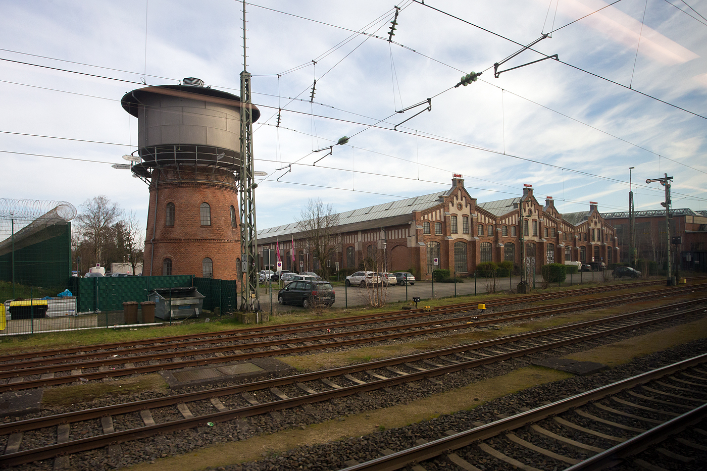 Ein Blick aus dem Zug im Bahnhof Lingen (Ems) am 14 M�rz 2024, auf das ehemalige Bundesbahn Ausbesserungswerk (AW). Links der Bahnwasserturm (es gibt auch einen h�heren St�dtischen Wasserturm in der N�he des ehemaligen Ausbesserungswerks), rechts davon die Halle 4, seit 1997 die gr��te Kunsthalle im westlichen Niedersachsen. Ganz rechts die vollst�ndig renovierten ehemaligen Lokrichthalle I/II, in denen sich heute die Hochschule Osnabr�ck, Campus Lingen befindet.

Der hohe Zaun mit dem S-Draht ganz links sichert nicht das Gel�nde der Kunsthalle, sondern links befindet sich die JVA Lingen (Justizvollzugsanstalt). 
