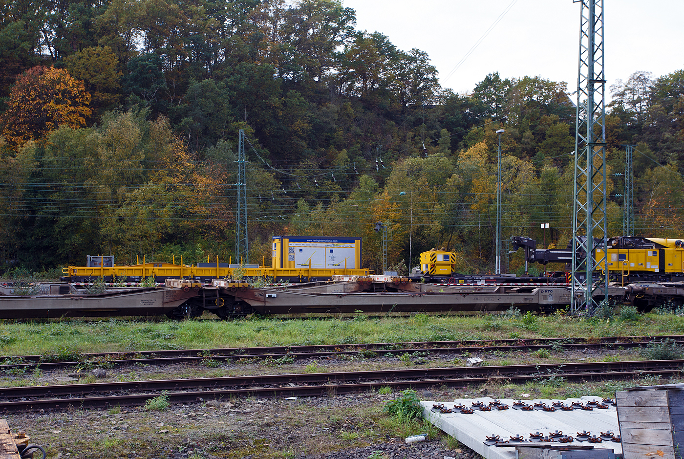 Ein Drehgestell-Flachwagen, Nummer unbekannt, der Gattung Res als Kran-Schutzwagen gegenlastseitig, u. a, beladen mit Werkstattcontainer, kleine Traversen, Anschlagmittel und sonstiges Zubehör/Equipment, sowie rechts der vierachsige Gegenlastwagen D-HGUI 99 80 9310 032-4 auf der einen Seite mit dem abgelegten Gegengewicht des 150 t Gleis- und Eisenbahndrehkran (EDK) KIROW Multi Tasker KRC 1200, D-HGUI 99 80 9419 041-5 „IDEFIX“ der HERING Bahnbau GmbH und auf der anderen Seite stützt, sich hier in der Transportstellung, das Gegengewicht-Teleskop hydraulisch auf dem Gegenlastwagen ab.

Abgestellt am 19 Oktober 2025 im Rbf Betzdorf/Sieg.