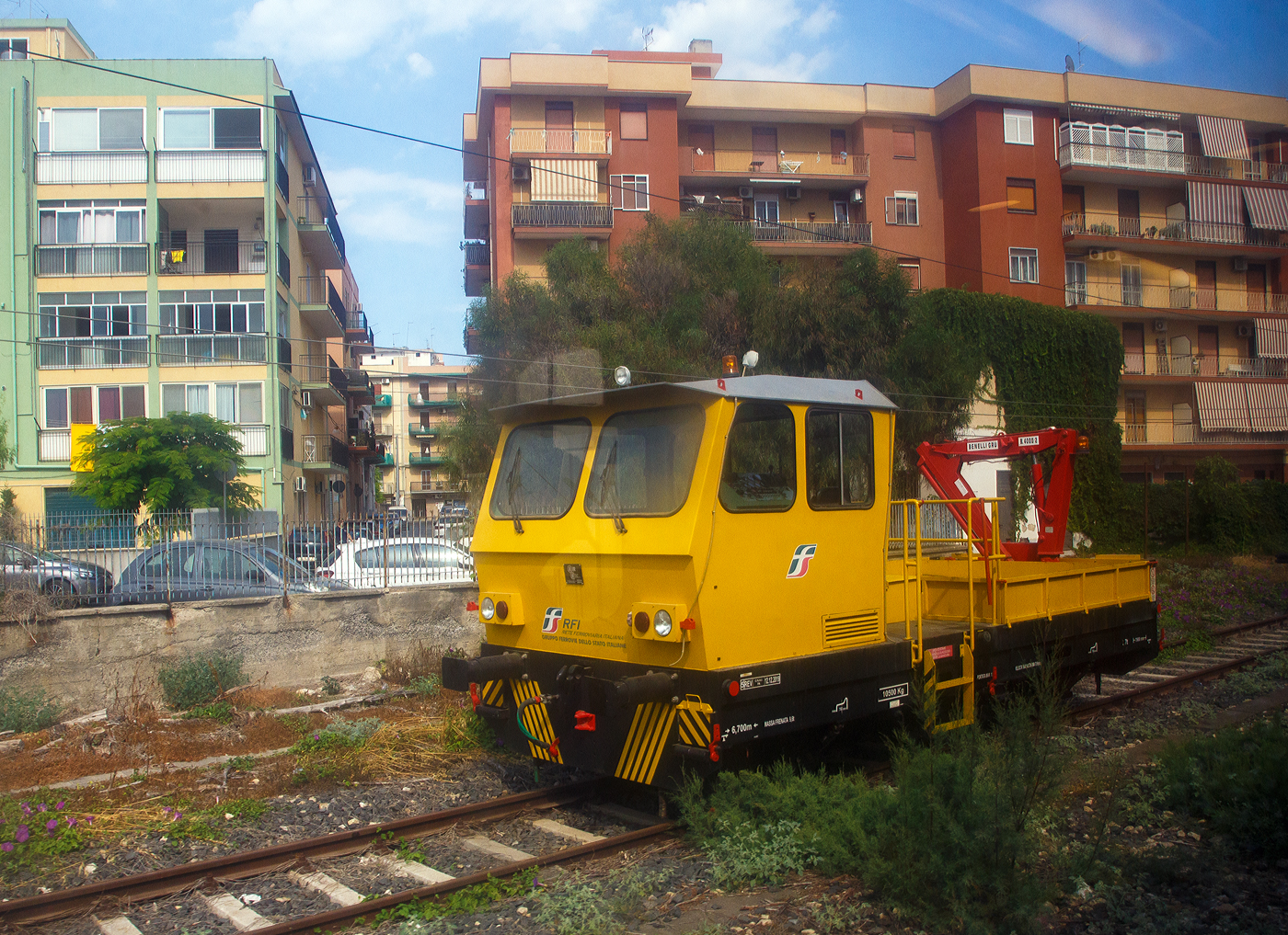 Ein Italienisches Gleisarbeitsfahrzeug mit hydraulischen Benelli Kran B.4000/2 (Autocarrello con gru vom Typ MERMEC MM 87) der RFI - Rete Ferroviaria Italiana (FS Eisenbahninfrastruktur) steht am 19 Juli 2022 bei Augusta (Sizilien). Da die Aufnahme aus einem Zug ist, leider mit Spieglungen. 