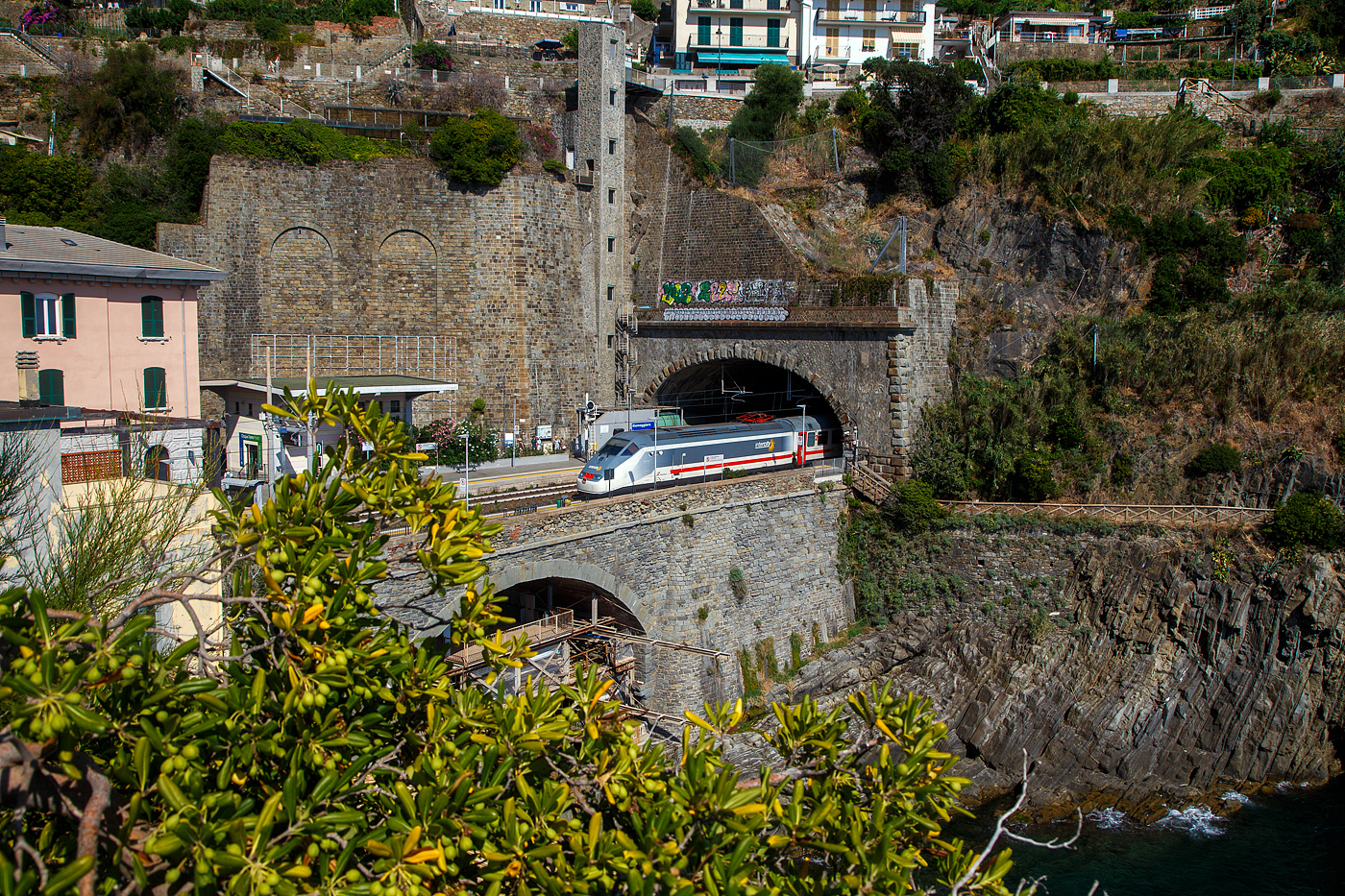 Ein Trenitalia Intercity (IC) rauscht im Sandwich zweier E.414 (ehemalige Triebköpfe E.404 A der ersten ETR 500 – Monotensione) durch den Cinque Terre Bahnhof Riomaggiore in Richtung Genua (Genova).

Vor dem Zuganfang auf der anderen Bahnsteigseite sieht am den Eingang von dem Fußgänger-Tunnel vom Bahnhof zum Zentrum von Riomaggiore. Dieser führt durch den Eisenbahntunnel, ist aber abgegrenzt. 

Zudem sieht man hinten auch den hohen Lift (Aufzug), mit diesem kommt man auf den etliche Meter oben liegenden Weg. Der Lift ist aber in der Saison kostenpflichtig, aber er erspart etliche Treppenstufen oder einen gewaltigen Umweg.