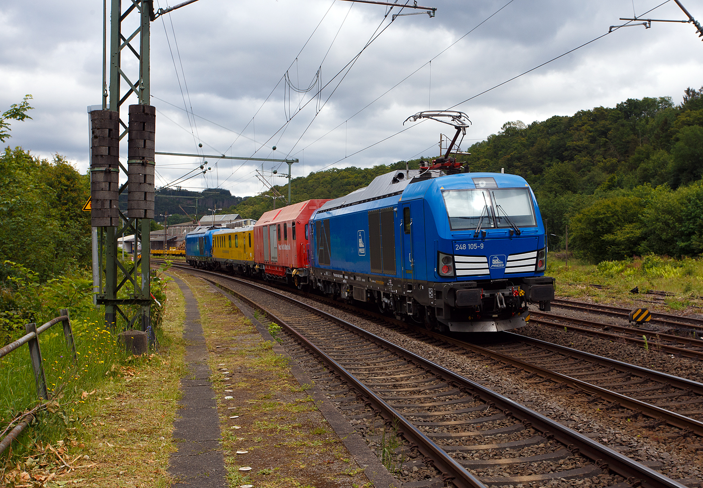 Im Sandwich zweier SIEMENS Vectron Dual Mode fährt ein Schienenprüfzug (SPZ 3), auf Prüffahrt (der Messwagen ist auf die Schiene absenkt) am 14 Juni 2024 durch Scheuerfeld/Sieg in Richtung Siegen.

Die vierteilige Komposition bestand aus:
• Der SIEMENS Vectron Dual Mode der 248 027-5 (90 80 2248 027-5 D-PRESS) alias 248 106-8 der PRESS (Eisenbahnbau- und Betriebsgesellschaft Pressnitztalbahn mbH, Jöhstadt).
• Dem Messwagen SPZ-STW (STW steht für Steuerwagen) 720 301(99 80 9360 006-7 D-DBMP) der DB InfraGO AG, ehemals DB Netz AG (NetzInstandhaltung/Fahrwegmessung).
• Einem Hilfszug-Wagen (Nummer unbekannt) der DB Netz AG (Notfalltechnik) bzw. DB InfraGO AG.
• Und der SIEMENS Vectron Dual Mode der 248 026-7 (90 80 2248 026-7 D-PRESS) alias 248 105-9 der PRESS (Eisenbahnbau- und Betriebsgesellschaft Pressnitztalbahn mbH, Jöhstadt).

Die Energieversorgung vom Messwagen und dem Hilfszug-Wagen erfolgte von den Loks (spartanisch) über außenverlegte Kabel.

Normalerweise ist der Schienenprüfzug ein zweiteiliger Messtriebwagen, bestehend aus Trieb- (ATW - 719 301) und einem Steuerwagen (STW - 720 301), siehe u.a. http://hellertal.startbilder.de/bild/Deutschland~Unternehmen~DB+Netz+AG/773623/der-plasser--theurer-schienenpruefzug-der.html Warum es nun diese Komposition ist, das ist mir nicht bekannt, vielleicht setzt man nun noch mehr auf E-antrieb.

Der Schienenprüfzug wurde 2015 von Plasser & Theurer in Linz (A) unter der Fabriknummer 6003 (SPZ-STW) gebaut. Um Schäden am Oberbau frühzeitig zu erkennen, setzt die Deutsche Bahn AG verschiedene Prüfzüge der Baureihen 719 und 720 ein. Mittels zweier zerstörungsfreier Prüfverfahren werden die Schienen auf Schädigungen untersucht. Bei der Ultraschallprüfung können Materialfehler durch Materialermüdung innerhalb der Schiene bis zum Schienenfuß erkannt werden. Mit der Wirbelstromprüfung wird der Schienenkopf geprüft.

Die beiden SIEMENS Vectron Dual Mode wurden 2022 von SIEMENS in München-Allach, unter den Fabriknummern 23168 und 23169, gebaut und an die PRESS - Eisenbahnbau- und Betriebsgesellschaft Pressnitztalbahn mbH in Jöhstadt geliefert. Die beiden Vectron DM haben die Zulassung in Deutschland für eine Höchstgeschwindigkeit 160 km/h.

Der Vectron Dual Mode vereint die Vorteile einer vollwertigen Diesellokomotive mit denen von elektrischen Lokomotiven. Dank des leistungsstarken Dieselantriebs sowie der Ausstattung für die Nutzung der Fahrleitung kann man jederzeit flexibel auf die Traktionsanforderungen reagieren. So bleibt man immer in Bewegung, egal ob mit oder ohne Fahrdraht. Wo ein Fahrdraht ist, sollte man ihn nutzen. Denn der Betrieb unter Oberleitung ist im Vergleich zum Dieselbetrieb deutlich kosteneffizienter und umweltfreundlicher. Doch weil es in Deutschland noch lange Zeit Strecken geben wird, die nicht vollständig elektrifiziert sind, fahren viele Diesellokomotiven unter Fahrdraht weiter. Das ist aber ökonomisch und ökologisch wenig sinnvoll. Dies ist hier ein Beispiel einer echten Alternative.
