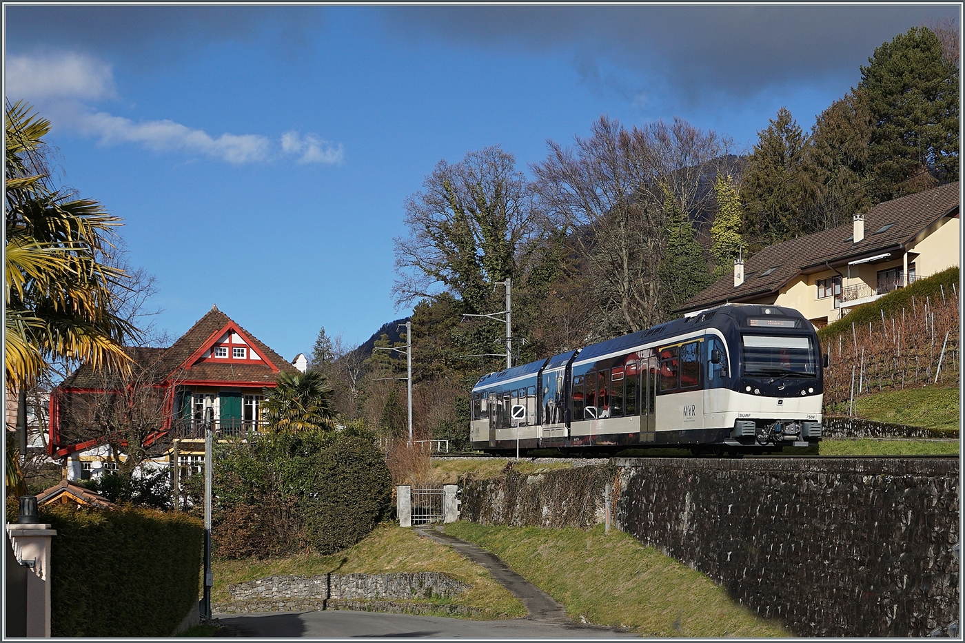 Kaum 19 Minuten nach der Abfahrt im verschneiten Les Avants erreicht der CEV MVR ABeh 2/6 7504  Vevey , der als R 34 2317 nach Montreux unterwegs ist, den Halt Planchamp (530 müM). Wie man unschwer feststellen kann ist der Zug nun in einer grünen Landschaft unterwegs, wobei bei genauerem Hinsehen sogar Plamen die Strecke säumen.

3. Januar 2025