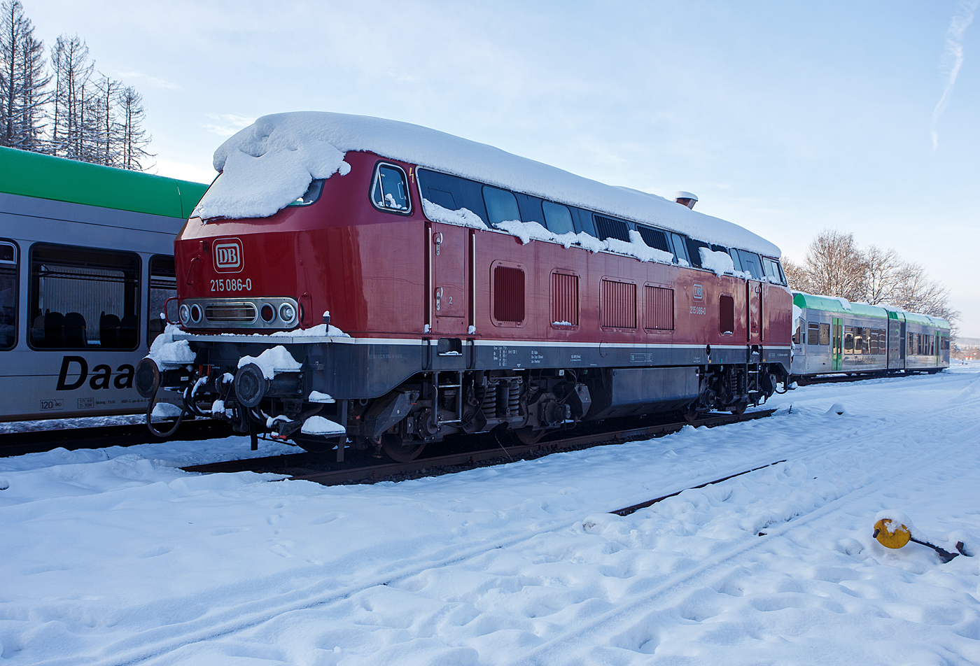 Nicht nur im Süden der Republik hat es etwas geschneit, wie hier auf dem Westerwald auf der Bindweide, aber nicht so mächtig.  Die für Steilstrecken zugelassenen 215 086-0 (eigentlich 225 086-8 / 92 80 1225 086-8 D-EVG) der RE - Rheinische Eisenbahn ist am ersten Advent  dem 3 Dezember 2023 auf dem Betriebshof der Westerwaldbahn (WEBA) auf der Bindweide bei Steinebach/Sieg abgestellt. Dahinter stehen noch die beiden Daadetalbahn STADLER GTW 2/6 VT 117 und VT 118 (646 417-5 und 646 418-3) der Westerwaldbahn (WEBA), ex VT 117 und ex VT 118 HellertalBahn, die beiden gingen nach Auflösung der HellertalBahn an die WEBA, der 3. der VT 116 ging an die HLB.

Die V 163 wurde 1971 bei MaK unter der Fabriknummer 2000091 und an die DB als 215 086-0 mit stärkerem Motor (1.840 kW) ausgeliefert und war bis 2000 bei der DB Regio AG. Im Jahr ging sie an die DB Cargo AG und wurde in die heutige 225 086-8 umgebaut und um gezeichnet.  Zum Umbau gehörte der Ausbau der Dampfheizung, da diese für Güterzüge nicht mehr benötigt wurde. Stattdessen wurde ein Vorwärm- und Warmhaltegerät für die Maschinenanlage eingebaut. Außerdem besitzen die Loks eine Doppeltraktionssteuerung, die Doppeltraktionen mit Loks der Baureihen 217 und 218 ermöglichen. 

Mit der Ausmusterung bei der nunmehrigen DB Schenker Rail Deutschland AG im Jahr 2011 erfolgte auch der Verkauf an die RE - Rheinische Eisenbahn, hier wurde sie wieder in den Auslieferzustand zurückversetzt und trägt nun die ursprüngliche Bezeichnung 215 086-0, ist aber eine 225er und hat so die NVR-Nummer 92 80 1225 086-8 D-EVG).

Die Lokomotiven der Baureihe 215 wurden kurzfristig als Variante der V160-Familie für den Personen- und Güterverkehr beschafft und dabei im Gegensatz zu anderen Baumustern der V160-Familie mit Heizdampferzeugern ausgerüstet. Geplant war sie im alten Baureihenschema als V 163. Diese Lokomotiven sollten später in die Baureihe 218 umgebaut werden können. Dazu erhielt sie bereits den um 400 Millimeter verlängerten Lokkasten anderer bis dahin nur als Prototyp vorhandener Baureihen. Der Hauptgrund für den Bau der Baureihe 215 lag darin, dass die Reihe 218 noch nicht Serienreife erreicht hatte, die Bundesbahn aber die restlichen noch vorhandenen Dampflokomotiven ersetzen wollte. 1968 bis 1970 wurden insgesamt 150 Lokomotiven gebaut.

Mitte der 1980er erhielten die Maschinen mit 12-Zylinder-V-Motor Abgashutzen auf dem Dach (wie diese). Sie bewirkten eine Beschleunigung und Umlenkung der Abgase, so dass bei klimatisierten Reisezugwagen, welche auf dem Dach die Luftansaugöffnungen hatten, eine Belästigung der Fahrgäste durch Abgase oder Abgasgerüche vermieden wurde. Weiterhin sorgten die Hutzen noch für andere Effekte: die bis zu 280 °C heißen Abgase wurden V-förmig an der Oberleitung vorbeigeleitet, so dass die Oberleitung nicht verrußt wurde und sich nicht zu sehr ausdehnte. Außerdem wurde die Gefahr von Böschungsbränden durch herabsinkende, heiße Rußpartikel reduziert. Auch eine Türschließanlage und Bremsgestängesteller zum automatischen Nachstellen des Bremskolbenhubes wurden nachgerüstet.

TECHNISCH DATEN:
Gebaute Stückzahl: 150
Spurweite:  1.435 mm (Normalspur)
Achsformel:  B'B'
Länge:  16.400 mm
Drehzapfenabstand:  8.600 mm
Achsabstand im Drehgestell:  2.800 m
Gesamtradstand: 11.400 mm
Gewicht: 79 Tonnen
Radsatzfahrmasse:  20,0 Tonnen
Höchstgeschwindigkeit: 140 km/h (90 km/h im Rangiergang) Motorentyp: MTU MA 12 V 956 TB 10 (ein V12-Zylinder-Diesel-Motor)
Dauerleistung: 1.840 kW (2.500 PS)
Nenndrehzahl: 1.500 min-1
Leistungsübertragung: hydraulisch, über Kardanwellen vom mittig angeordneten Getriebe zu den beiden zweiachsigen Drehgestellen. So werden über Radsatzgetriebe jeweils beide und somit alle Achsen angetrieben. Ein Wendegetriebe ermöglicht den Richtungswechsel.
Getriebe: Voith L 820 brs  
Tankinhalt: 3.050 l (Kraftstoff) 
Sandvorrad: 320 kg 
Sie ist für Steilstrecken zugelassen.
