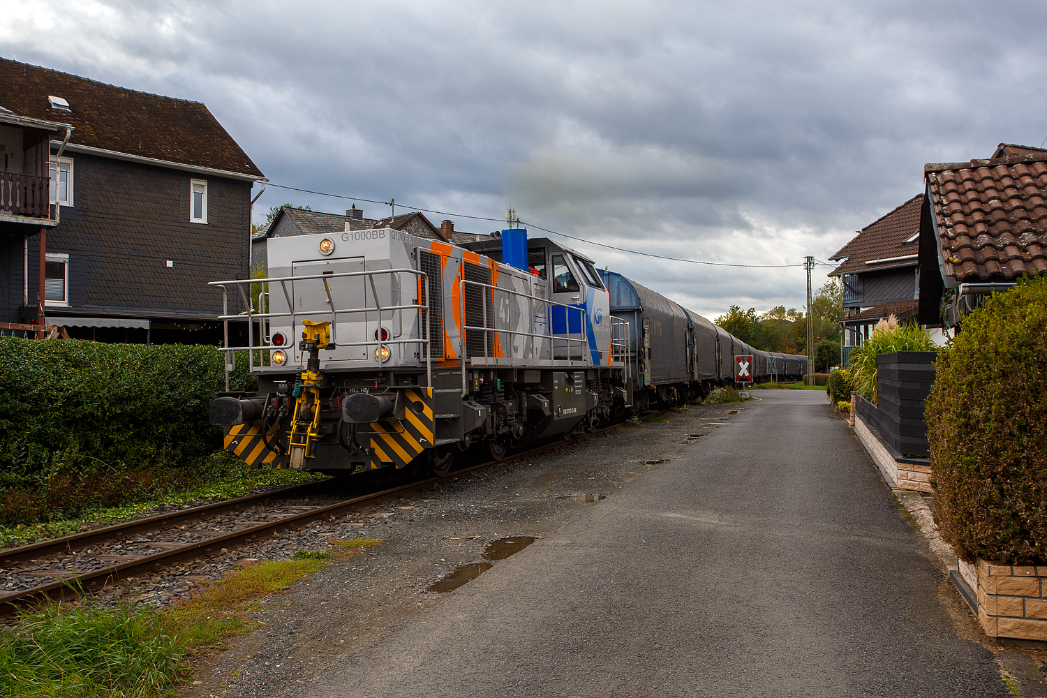 Nun hat die KSW 47 (92 80 1271 027-5 D-KSW), ex D 2 der HFM, eine Vossloh G 1000 BB der KSW (Kreisbahn Siegen-Wittgenstein), den Coilzug übernommen und zieht diesen nun vom KSW Rbf Herdorf weiter zu den Schäfer Werke KG am Pfannenberg in Neunkirchen-Salchendorf. Hier geht es gerade in Neunkirchen-Struthütten am 18 September 2025 zwischen den Häuser und Hecken hindurch. 

Auch wenn hierdurch, mehrmals täglich, die Anwohner genervt werden, zu Einem durch die Rollgeräusche und vor allem durch das erforderliche Pfeifen (Hupen) der Loks, wegen der vielen Bahnübergänge. Aber dennoch ist es schon lange praktifizierter Umweltschutz, denn mit allein dieser Zug ersetzt 40 bis 50 LKW. Übrigens diese Bahnstrecke der ehemaligen Freien Grunder Eisenbahn AG (auch bekannt als „Talbahn“) gibt es seit 1904, heute gehört sie als Betriebsstätte FGE -Freien Grunder Eisenbahn zur KSW Kreisbahn Siegen-Wittgenstein GmbH und wird als Freien Grunder Eisenbahn NE 447 (DB - Streckennummer 9275) geführt.

Mit einer eigenen Schieneninfrastruktur im Kreis Siegen-Wittgenstein, die mehr als 30 Kilometer umfasst, verfügt die KSW über wichtige Kapazitäten im wachsenden Güterverkehr. Auch wenn in Deutschland der Schienen-Güterverkehr geringer geworden ist, so ist er hier auf der „Talbahn“ die letzten Jahre ungemein gewachsen. 

Früher gab es ich Siegen (Eintracht) noch einen Abschnitt, wo die KSW bzw. frühere Siegener Kreisbahn auf der Eiserfelder Straße mit Läute (Glocke) fuhr, so wurde diese Bahn auch gerne im Volksmund „Bimmelbahn“ genannt.